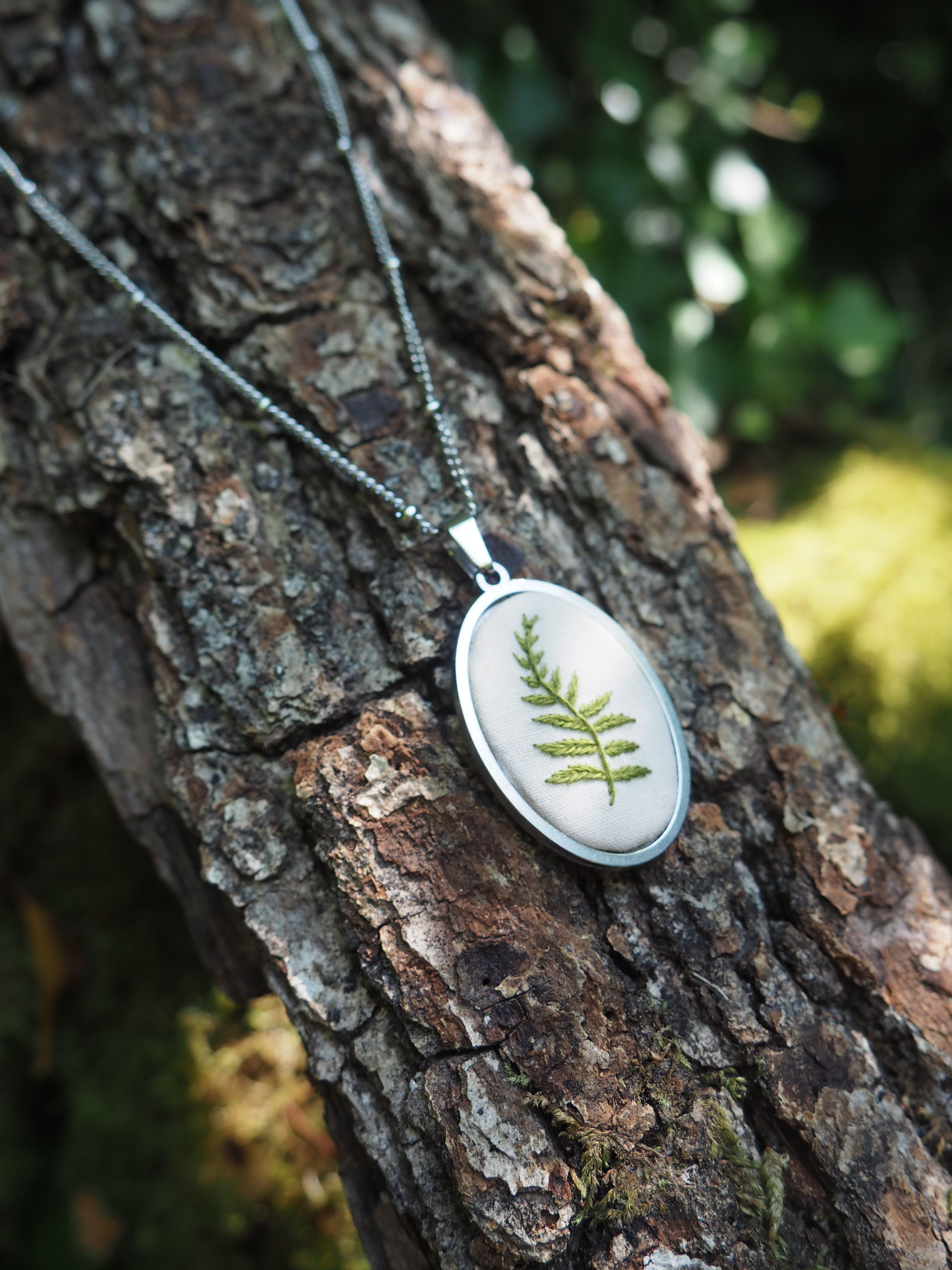 Cette image montre un délicat pendentif posé sur l'écorce d'un arbre. Le pendentif est de forme ovale avec un cadre en argent, et contient une véritable feuille de fougère préservée sous ce qui semble être une résine transparente. Il est attaché à une fine chaîne argentée. La photo est prise en gros plan avec un bel effet de profondeur de champ, l'arrière-plan verdoyant étant légèrement flou. L'écorce rugueuse et texturée de l'arbre crée un contraste intéressant avec la finesse et la délicatesse du bijou. C'est une belle composition qui met en valeur cette pièce de bijouterie naturelle et artisanale.