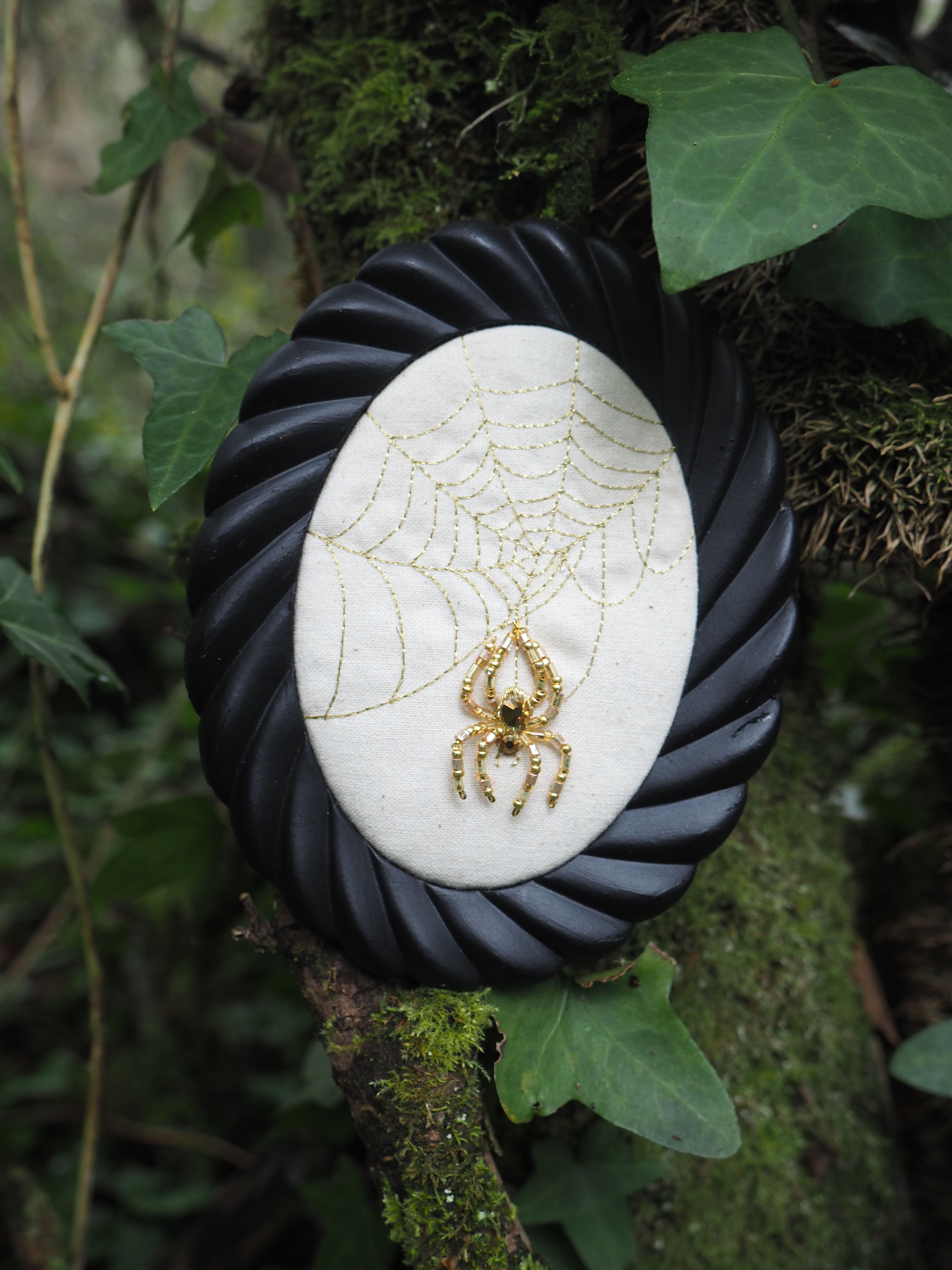 L'image présente un cadre ovale noir avec un motif torsadé, suspendu ou posé sur un tronc d'arbre couvert de mousse et de lierre. À l'intérieur du cadre, sur un fond de tissu blanc cassé, est brodée une toile d'araignée dorée. Au centre de cette toile, une broche en forme d'araignée dorée, ornée de ce qui semble être une petite pierre noire ou un cabochon au niveau de son corps, est épinglée. L'arrière-plan est flou, mais on distingue clairement la texture du tronc d'arbre, la mousse verte et les feuilles de lierre qui encadrent le sujet principal. L'éclairage semble naturel, mettant en valeur les détails dorés de l'araignée et de la toile. L'ambiance générale est un mélange de nature et d'un élément décoratif, avec une touche légèrement mystérieuse ou fantaisiste due au thème de l'araignée.