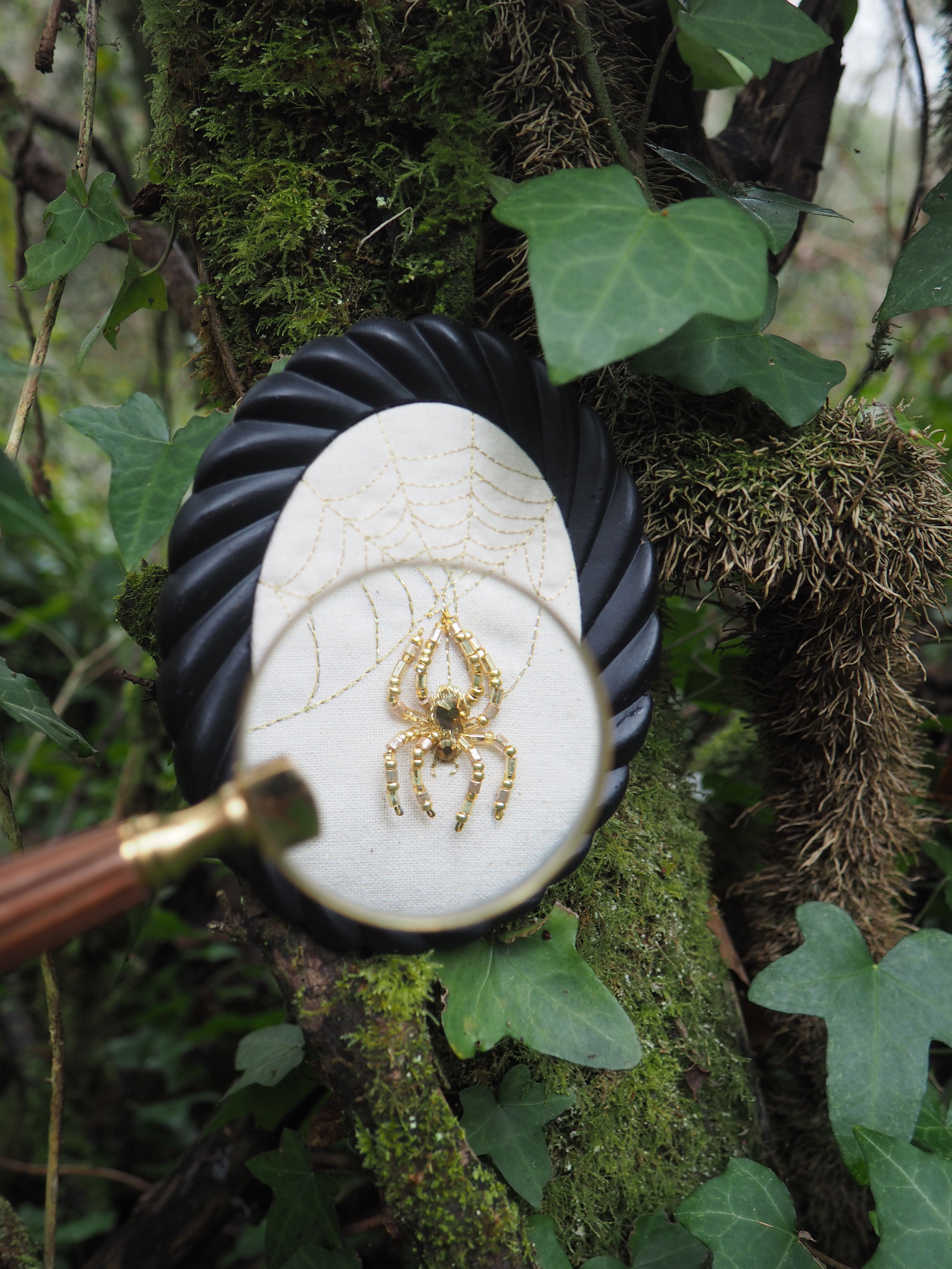 Cette image présente une scène intrigante et détaillée, centrée sur une broderie d'araignée observée à travers une loupe, le tout placé dans un environnement naturel. Au premier plan, une loupe est tenue, son manche en bois et en laiton s'étendant vers le bas à gauche. La lentille de la loupe est positionnée de manière à agrandir une partie de la broderie. Au centre de l'image, derrière la loupe, se trouve un petit cadre ovale noir, avec un motif en spirale ou torsadé sur son pourtour. À l'intérieur de ce cadre, une pièce de tissu blanc est visible. Sur ce tissu, une broderie délicate représente une toile d'araignée, avec des fils dorés ou de couleur claire formant les rayons et les spirales de la toile. Au centre de cette toile, une araignée est brodée avec des perles dorées et des pierres vertes, lui donnant un aspect précieux et détaillé. L'araignée est représentée avec ses huit pattes et son corps segmenté. L'arrière-plan est composé d'un tronc d'arbre ou d'une branche épaisse, recouverte de mousse verte et de lierre. Les feuilles de lierre, d'un vert profond, sont visibles à plusieurs endroits, ajoutant à l'ambiance naturelle et un peu mystérieuse de la scène. La texture de l'écorce de l'arbre est rugueuse et irrégulière, contrastant avec la finesse de la broderie. La lumière semble naturelle et diffuse, éclairant la scène de manière douce. L'ensemble de l'image suggère une exploration minutieuse de la nature et de l'artisanat, avec un focus particulier sur les détails.