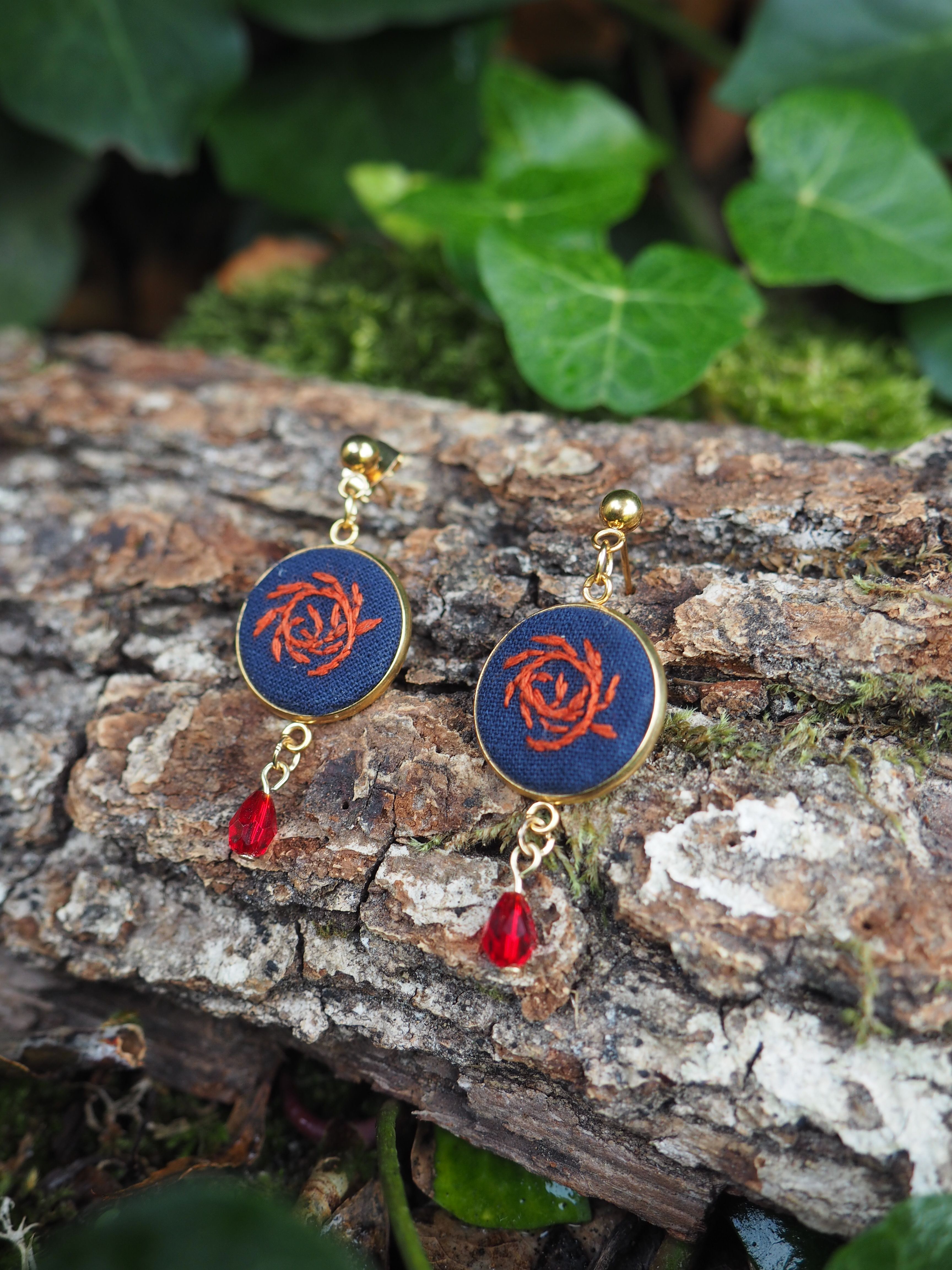 Cette image présente une paire de boucles d'oreilles élégantes posées sur un tronc d'arbre dans un environnement naturel.  **Description des bijoux :**
- Les boucles d'oreilles comportent des pendentifs circulaires de couleur bleu marine ornés d'un motif de rose rouge au centre
- Elles sont complétées par de petites perles rouges suspendues en dessous
- Les attaches et éléments de liaison semblent être dorés, créant un joli contraste **Cadre naturel :**
- Les bijoux reposent sur l'écorce rugueuse et texturée d'un tronc d'arbre
- L'arrière-plan est composé de feuillage vert luxuriant, légèrement flou
- L'éclairage naturel met en valeur les couleurs vives des boucles d'oreilles Cette mise en scène crée un contraste artistique intéressant entre l'élégance raffinée des bijoux et la rusticité naturelle de l'environnement forestier. Les couleurs - bleu, rouge et doré - ressortent particulièrement bien sur le fond naturel de bois et de verdure.