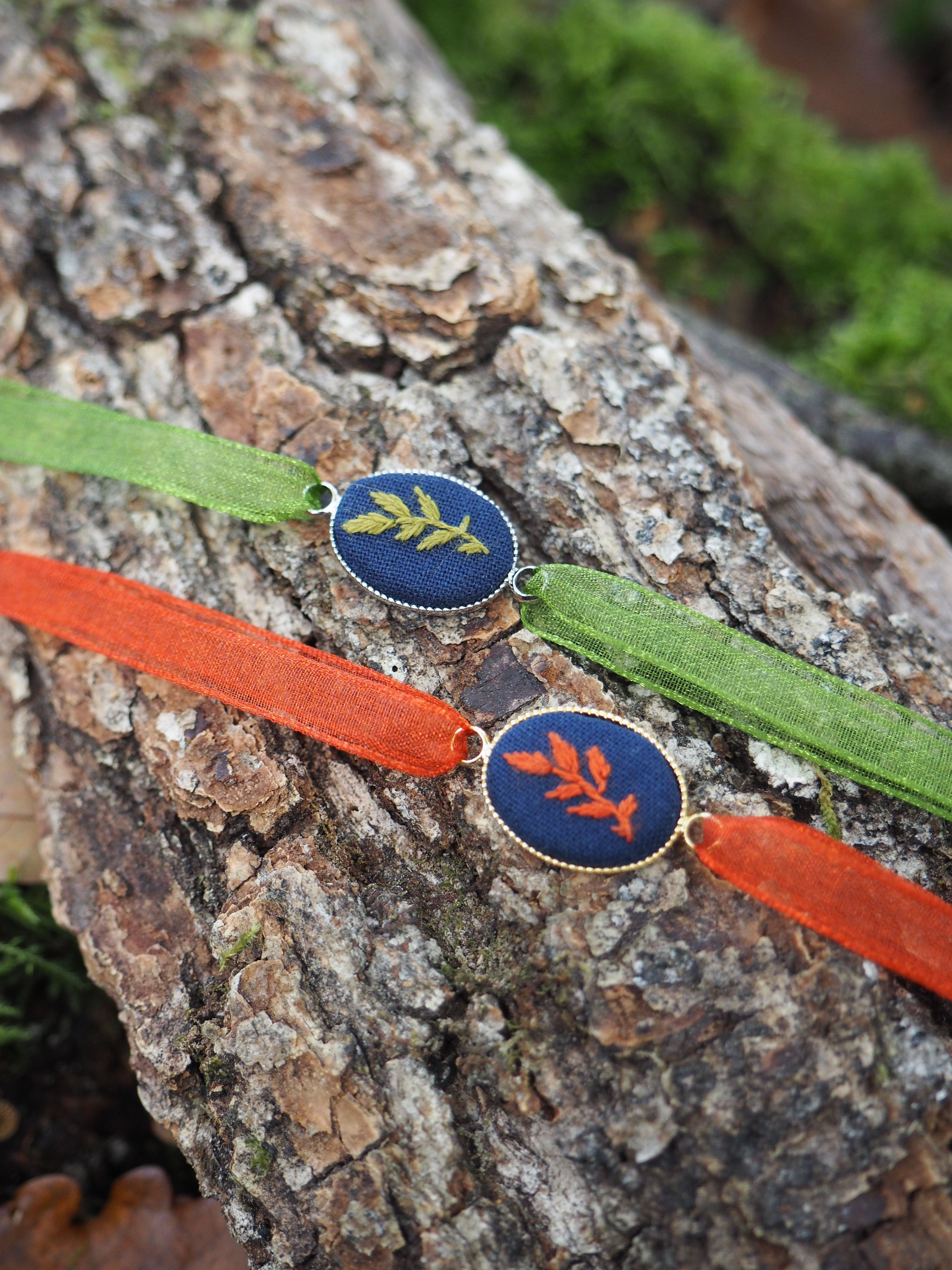 Cette image présente deux bracelets élégants posés sur un tronc d'arbre à l'écorce rugueuse et texturée.  **Les bracelets :**
- **Bracelet vert** : Orné d'un médaillon circulaire bleu marine avec un motif doré représentant une feuille ou une branche stylisée
- **Bracelet orange/rouge** : Également équipé d'un médaillon bleu marine, mais avec un motif doré différent évoquant une forme florale ou végétale **L'environnement :**
- Support naturel : un tronc d'arbre avec une écorce brune, grise et beige aux multiples nuances et aspérités
- Arrière-plan : végétation verte floue créant un bokeh naturel qui met en valeur les bijoux
- Éclairage : lumière naturelle douce qui fait ressortir les détails des médaillons dorés **Style et esthétique :**
Ces bijoux semblent s'inspirer de la nature avec leurs motifs botaniques dorés sur fond bleu. Le contraste entre les couleurs vives des bracelets (vert et orange) et le cadre naturel brun de l'écorce crée une composition harmonieuse qui évoque un style artisanal ou éco-responsable.