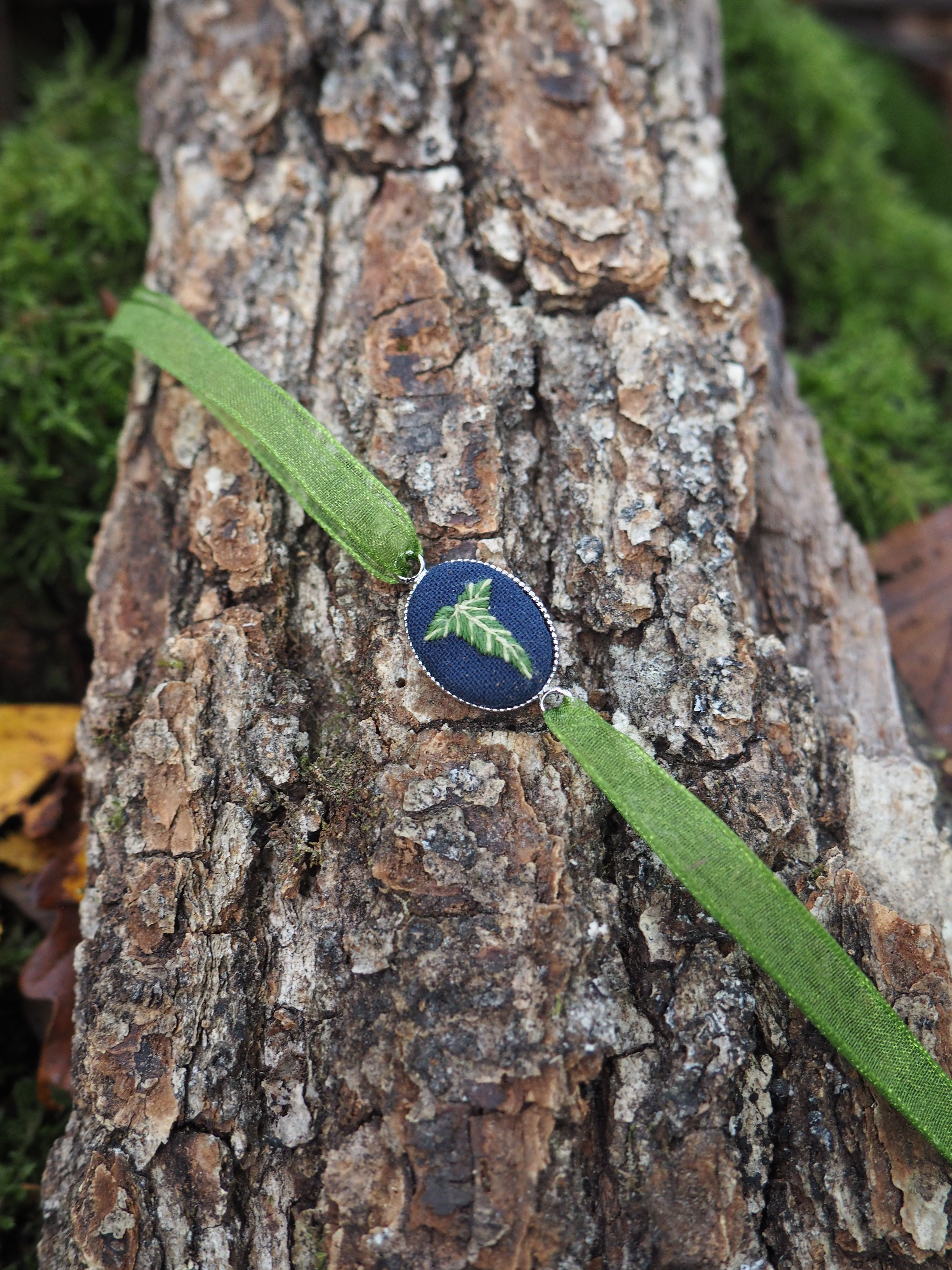 Cette image présente une composition naturelle très esthétique. Au centre, on observe un petit pendentif ou bijou de forme ovale avec une surface bleu-vert iridescente qui rappelle les couleurs d'une aile de papillon ou d'un scarabée. Ce bijou est délicatement posé sur un ruban ou une bande de tissu vert olive qui traverse diagonalement l'image. L'ensemble repose sur un tronc d'arbre ou une branche dont l'écorce rugueuse et texturée présente des tons bruns et gris, avec des motifs naturels créés par les fissures et les reliefs du bois. En arrière-plan, on distingue un tapis de mousse d'un vert vif qui apporte une belle profondeur de champ et fait ressortir les éléments du premier plan. La photographie capture bien les contrastes entre les différentes textures - la douceur du tissu, la rugosité de l'écorce et le velouté de la mousse - ainsi que la palette de couleurs naturelles dominée par les verts et les bruns, avec cette touche d'éclat iridescent du bijou qui attire immédiatement le regard.