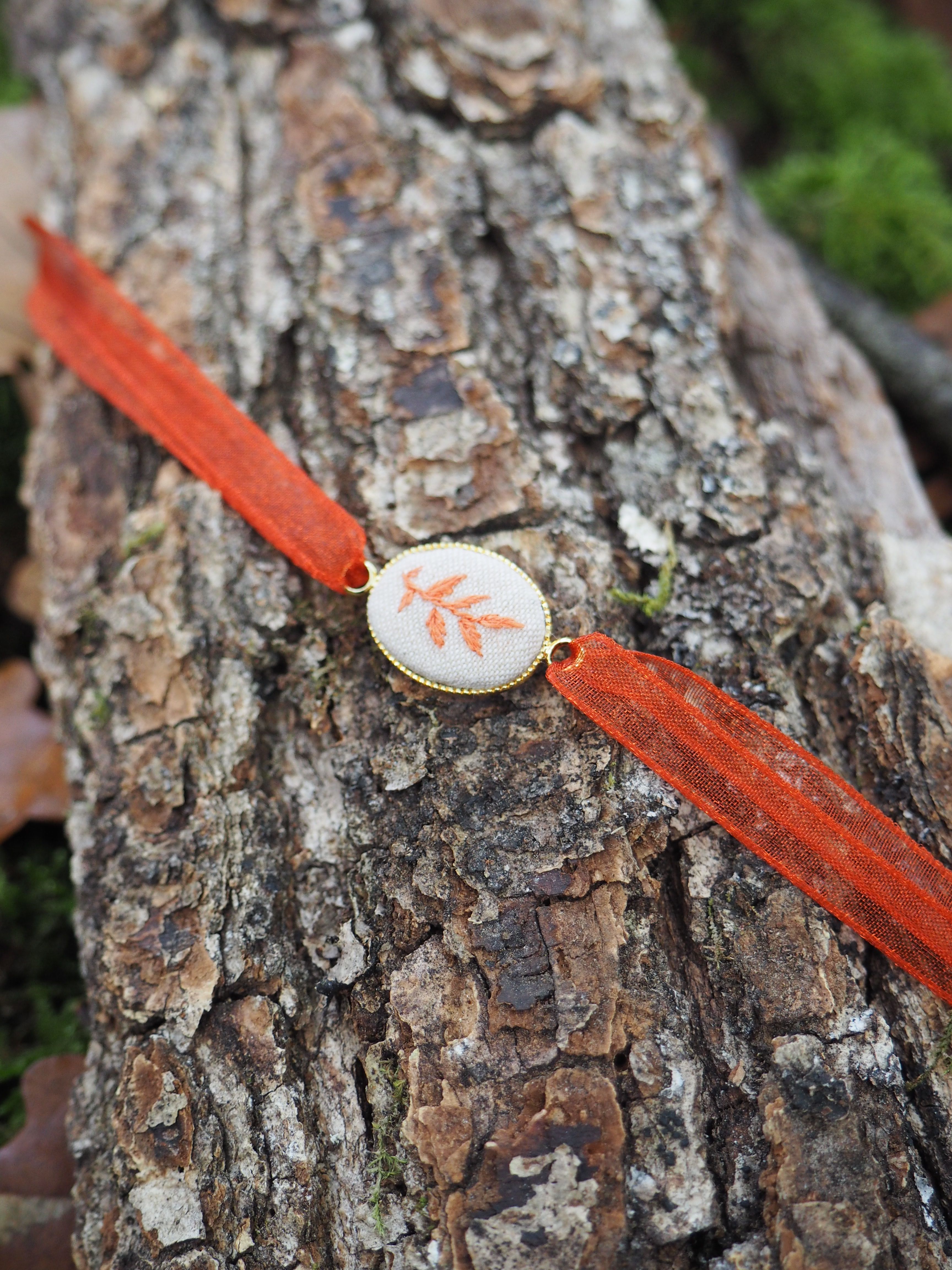 Cette image montre un bracelet ou un accessoire décoratif posé sur l'écorce rugueuse d'un tronc d'arbre. Le bracelet présente les caractéristiques suivantes : **Éléments principaux :**
- Un ruban ou cordon de couleur orange/rouge vif
- Un médaillon central rond de couleur blanche ou crème
- Sur le médaillon, on distingue un motif décoratif qui ressemble à une feuille ou un motif floral de couleur orange/corail **Contexte :**
- L'objet est photographié sur l'écorce naturelle d'un arbre, avec sa texture caractéristique rugueuse et ses tons bruns-gris
- L'arrière-plan est légèrement flou, laissant apercevoir de la végétation verte
- L'éclairage naturel met en valeur les contrastes entre les couleurs vives du bracelet et les tons neutres de l'écorce **Style :**
L'ensemble évoque un accessoire artisanal ou fait main, avec un style naturel et bohème qui s'harmonise bien avec l'environnement forestier dans lequel il est photographié.