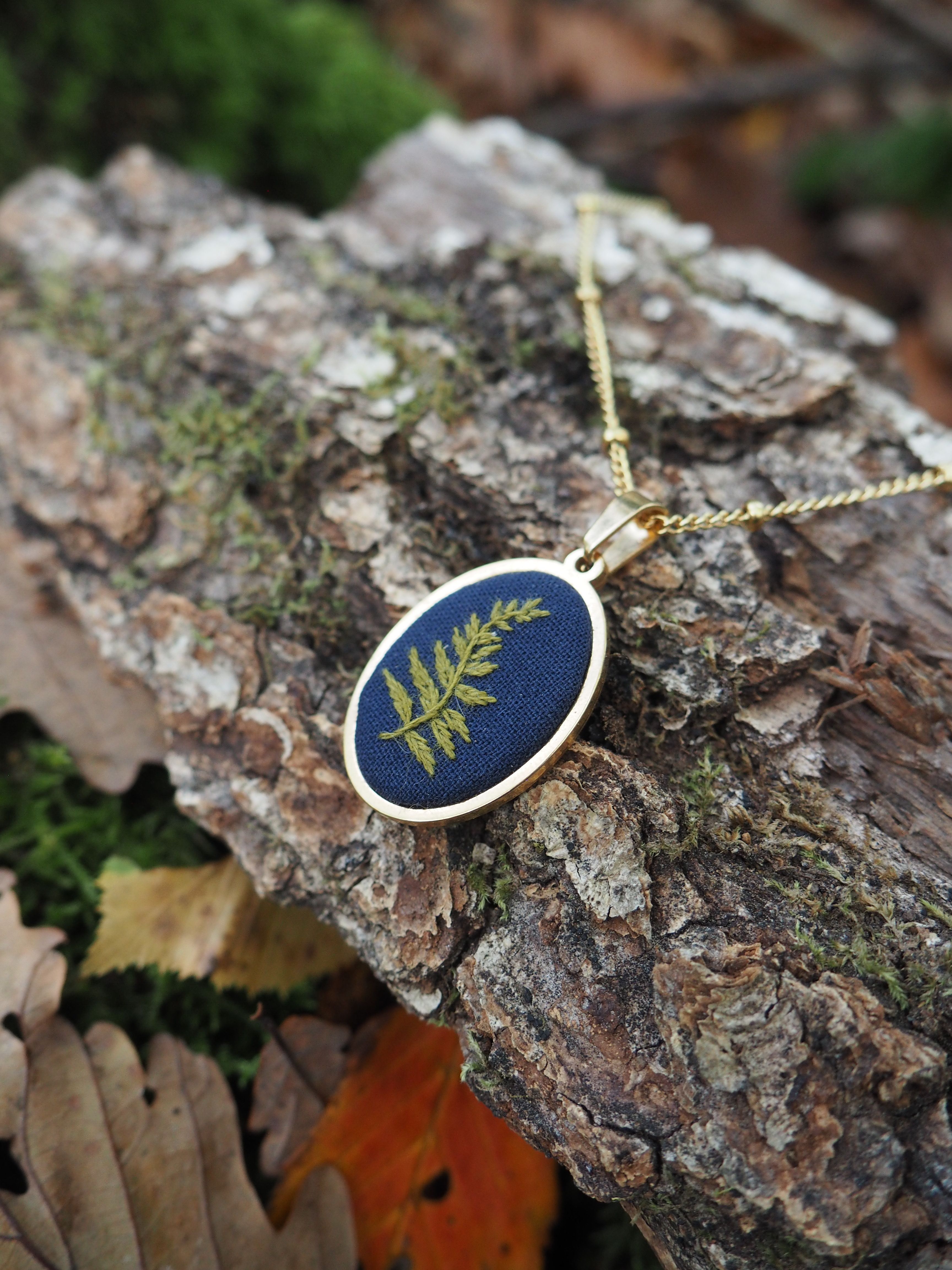 Cette image présente un bijou artisanal posé sur un tronc d'arbre dans un environnement naturel. Il s'agit d'un pendentif de forme ovale avec un cadre doré qui contient une véritable fougère séchée et préservée sur un fond bleu marine. Le pendentif est suspendu à une chaîne dorée délicate. Le bijou repose sur l'écorce rugueuse et texturée d'un tronc d'arbre, entouré de feuilles d'automne aux couleurs chaudes - des tons orangés, bruns et dorés. On peut également apercevoir de la mousse verte sur l'écorce, ainsi que de la végétation floue en arrière-plan, créant une atmosphère naturelle et organique. Ce type de bijou s'inscrit dans la tendance des créations botaniques où de véritables éléments végétaux sont incorporés dans des pièces de joaillerie, créant un lien direct avec la nature. La photographie met en valeur le contraste entre l'élégance du bijou et la rusticité de son environnement naturel.