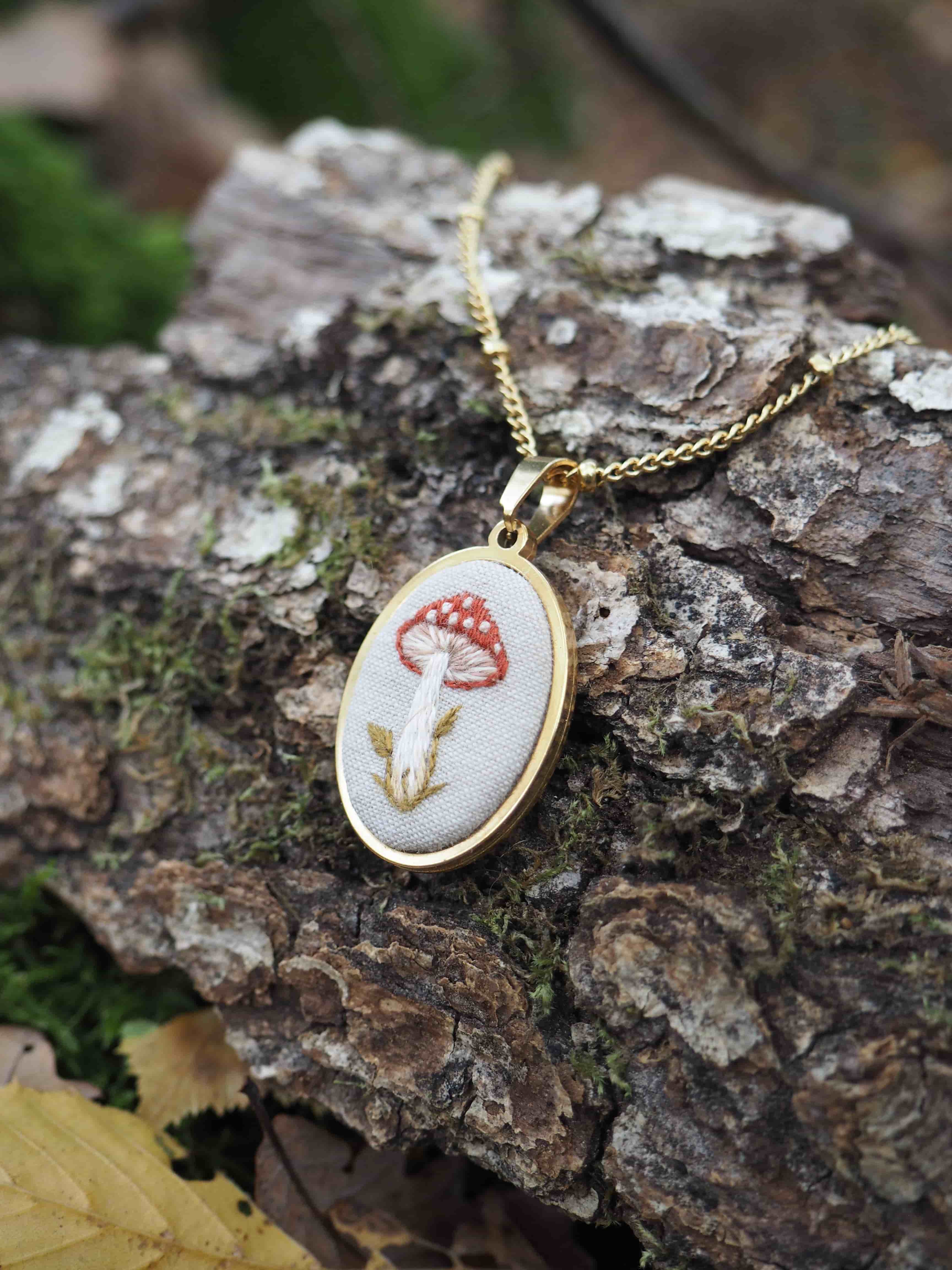 Cette image présente un délicat collier posé sur un morceau d'écorce d'arbre dans un environnement naturel. Le pendentif est de forme ovale avec un cadre doré élégant, et il contient une broderie ou un motif textile représentant un champignon rouge à pois blancs - très probablement une amanite tue-mouches avec sa couleur rouge caractéristique et ses taches blanches distinctives. Le champignon est accompagné de ce qui semble être une petite tige ou branche brodée en fil doré ou beige. L'ensemble du motif est réalisé sur un fond de tissu blanc ou crème, créant un contraste harmonieux avec les couleurs vives du champignon. Le collier repose sur une surface d'écorce rugueuse et texturée, avec de la mousse verte visible, ce qui crée une mise en scène très naturelle et poétique. L'arrière-plan est artistiquement flouté, mettant en valeur le bijou comme point focal de l'image. Ce type de bijou évoque un style artisanal, peut-être fait main, avec une esthétique qui rappelle les contes de fées et la nature sauvage.