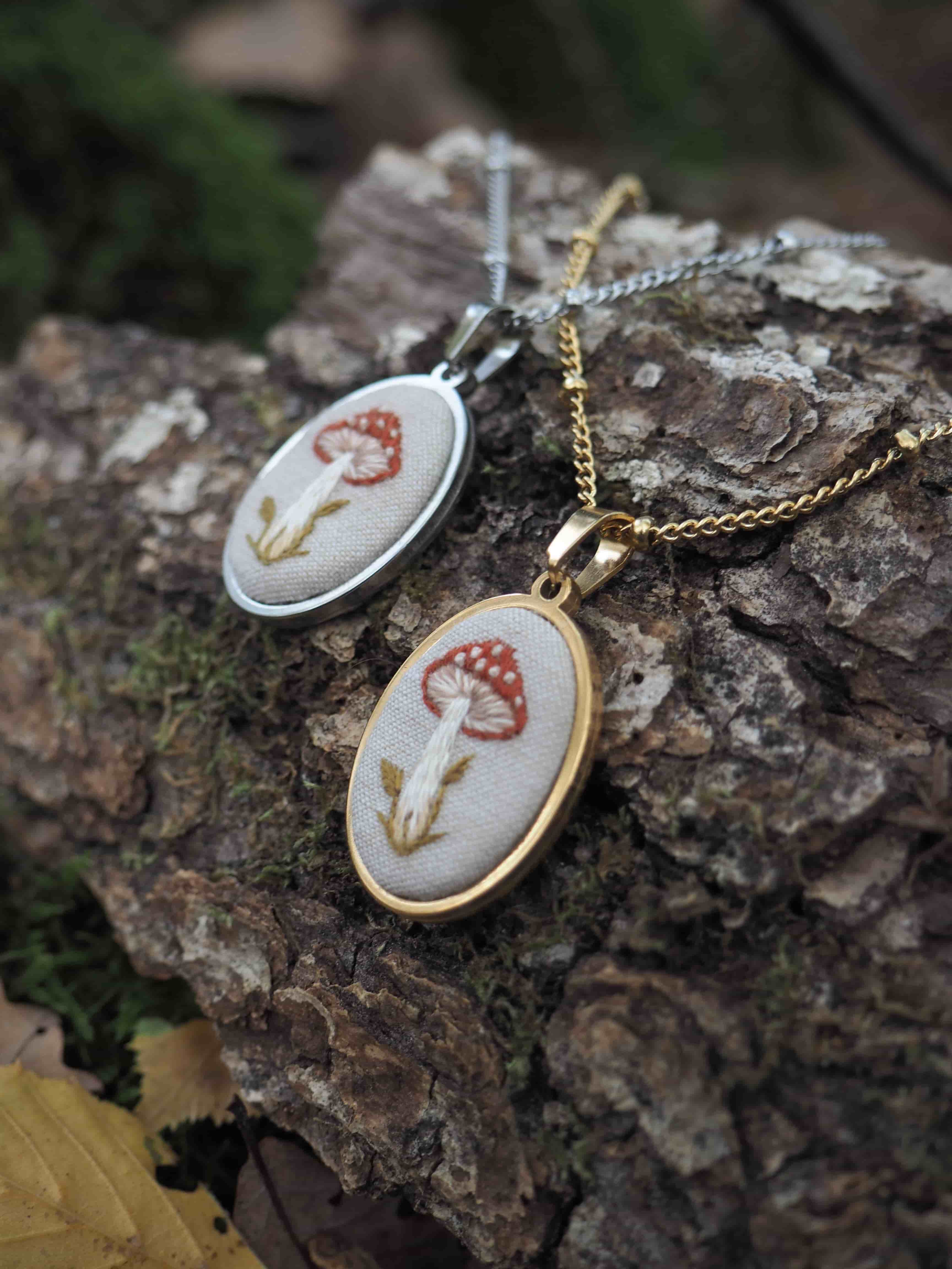 Cette image présente deux pendentifs délicats posés sur une surface en bois naturel dans un environnement extérieur.  **Caractéristiques des bijoux :**
- Deux pendentifs de forme ovale avec des montures métalliques (une dorée, une argentée)
- Chacun arbore le même motif brodé : un champignon rouge à pois blancs (amanite tue-mouches) avec une tige beige/marron
- La broderie est réalisée sur un fond de tissu blanc ou crème
- Les chaînes sont assorties aux montures respectives **Composition et ambiance :**
- Les pendentifs reposent sur une écorce d'arbre texturée, créant un contraste naturel
- L'arrière-plan est artistiquement flouté, révélant une végétation verte
- L'éclairage naturel met en valeur les détails fins de la broderie
- La mise en scène évoque un style bohème ou artisanal, en harmonie avec le thème champêtre des motifs Cette photographie met en avant le savoir-faire artisanal de ces bijoux brodés, probablement faits main, dans un cadre naturel qui complète parfaitement leur esthétique inspirée de la nature.