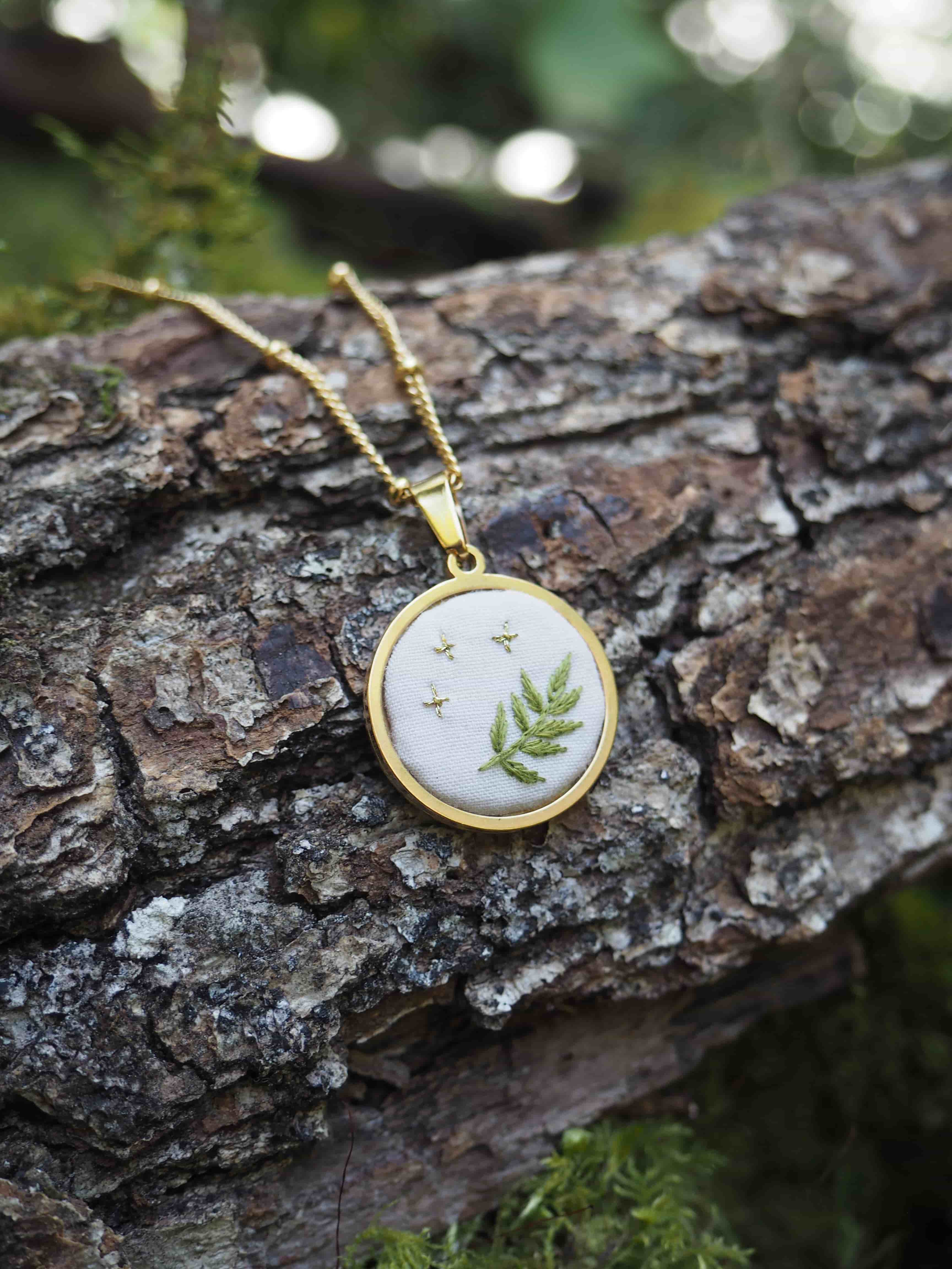 Cette image montre un délicat pendentif ovale sur une chaîne dorée, photographié sur un tronc d'arbre texturé. Le pendentif contient ce qui semble être une vraie feuille de fougère pressée et conservée sous verre, avec un cadre doré. L'arrière-plan est flou mais on peut distinguer des tons verts naturels, créant une belle harmonie avec l'écorce rugueuse de l'arbre. Le bijou a un aspect très naturel et botanique, représentant parfaitement la fusion entre la nature et l'artisanat. La composition de la photo met bien en valeur le contraste entre la délicatesse du pendentif et la texture brute de l'écorce.