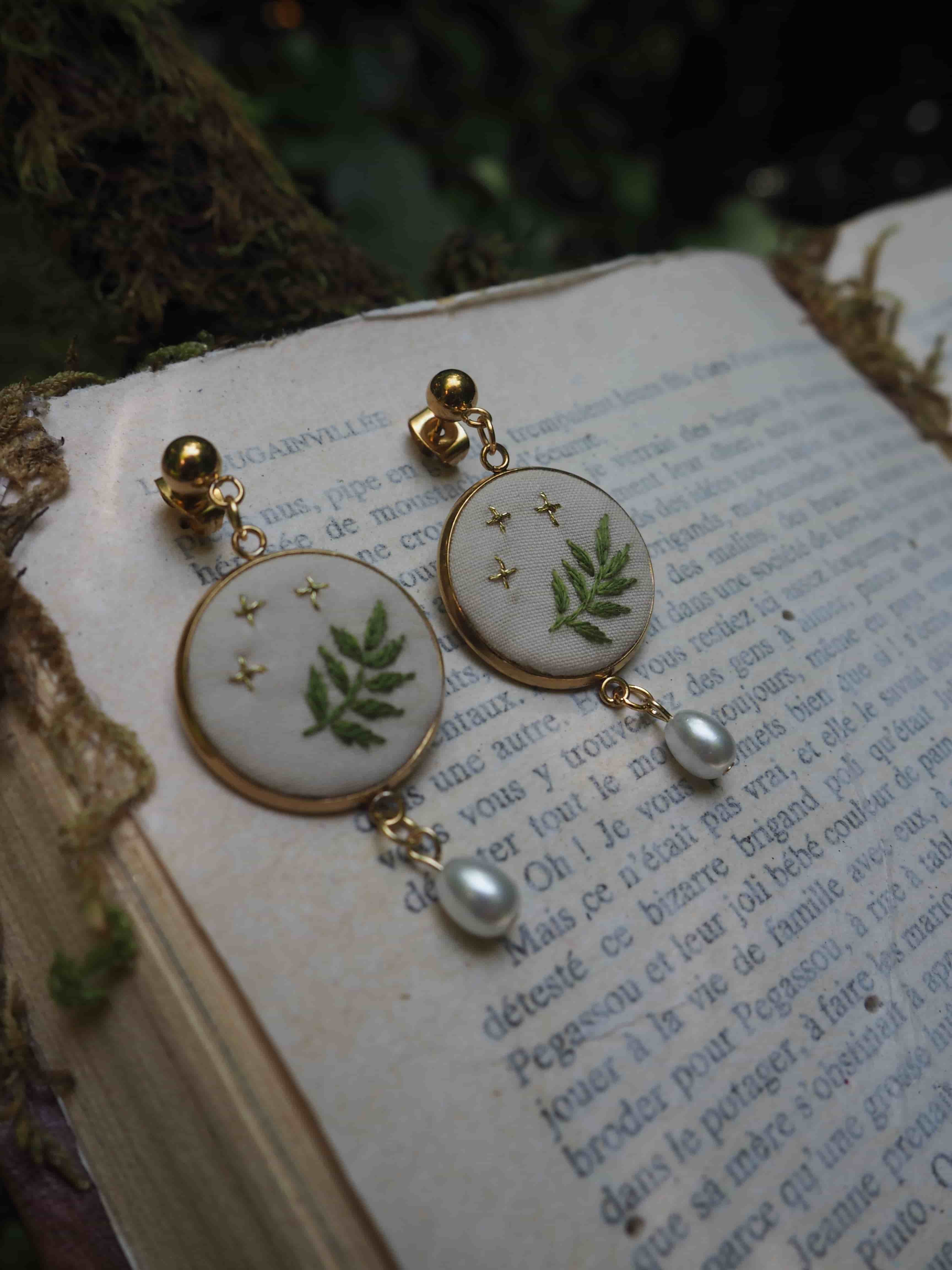 L'image montre une paire de boucles d'oreilles élégantes posées sur un livre ouvert. Les boucles d'oreilles sont composées de médaillons ronds blancs décorés de motifs de fougères vertes et de petites étoiles dorées. Au bas de chaque médaillon pend une petite perle nacrée. Les boucles d'oreilles semblent être montées avec des clips ou des tiges dorées. L'arrière-plan montre les pages d'un livre ancien avec du texte en français, donnant à l'image une ambiance vintage et romantique. La composition est artistique, avec un éclairage doux et naturel qui met en valeur les détails délicats des bijoux. Ces boucles d'oreilles ont un style botanique et délicat qui évoque la nature et le printemps, avec leur design incorporant des éléments naturels comme les fougères.