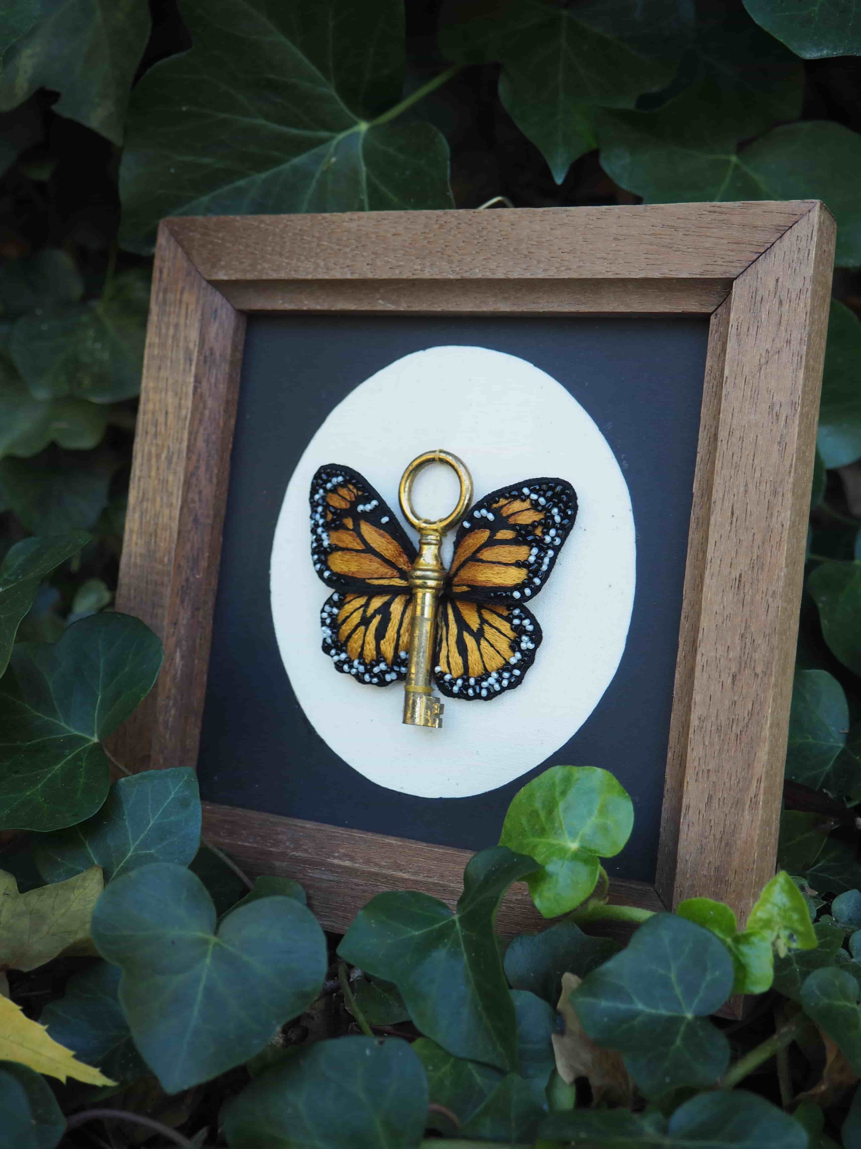 Cette image présente une œuvre d'art décorative encadrée, mise en scène dans un environnement naturel. Voici sa description détaillée : **L'œuvre principale :**
- Un cadre en bois de couleur naturelle/brune contient une composition artistique
- Le fond est noir, créant un contraste saisissant
- Au centre, on trouve un cercle blanc sur lequel est représenté un papillon monarque aux couleurs orange et noires caractéristiques
- Une clé dorée/dorée est artistiquement intégrée à la composition, semblant traverser ou être attachée au papillon **La mise en scène :**
- Le cadre est posé au milieu d'une végétation luxuriante
- On aperçoit de grandes feuilles vertes qui entourent complètement l'œuvre
- La végétation semble être celle d'une plante grimpante ou rampante, avec des feuilles en forme de cœur **L'effet artistique :**
Cette composition crée un dialogue poétique entre l'art et la nature, où le papillon encadré semble faire écho à l'environnement naturel qui l'entoure. La clé ajoute une dimension symbolique, évoquant peut-être l'idée de transformation ou d'accès à la beauté naturelle.