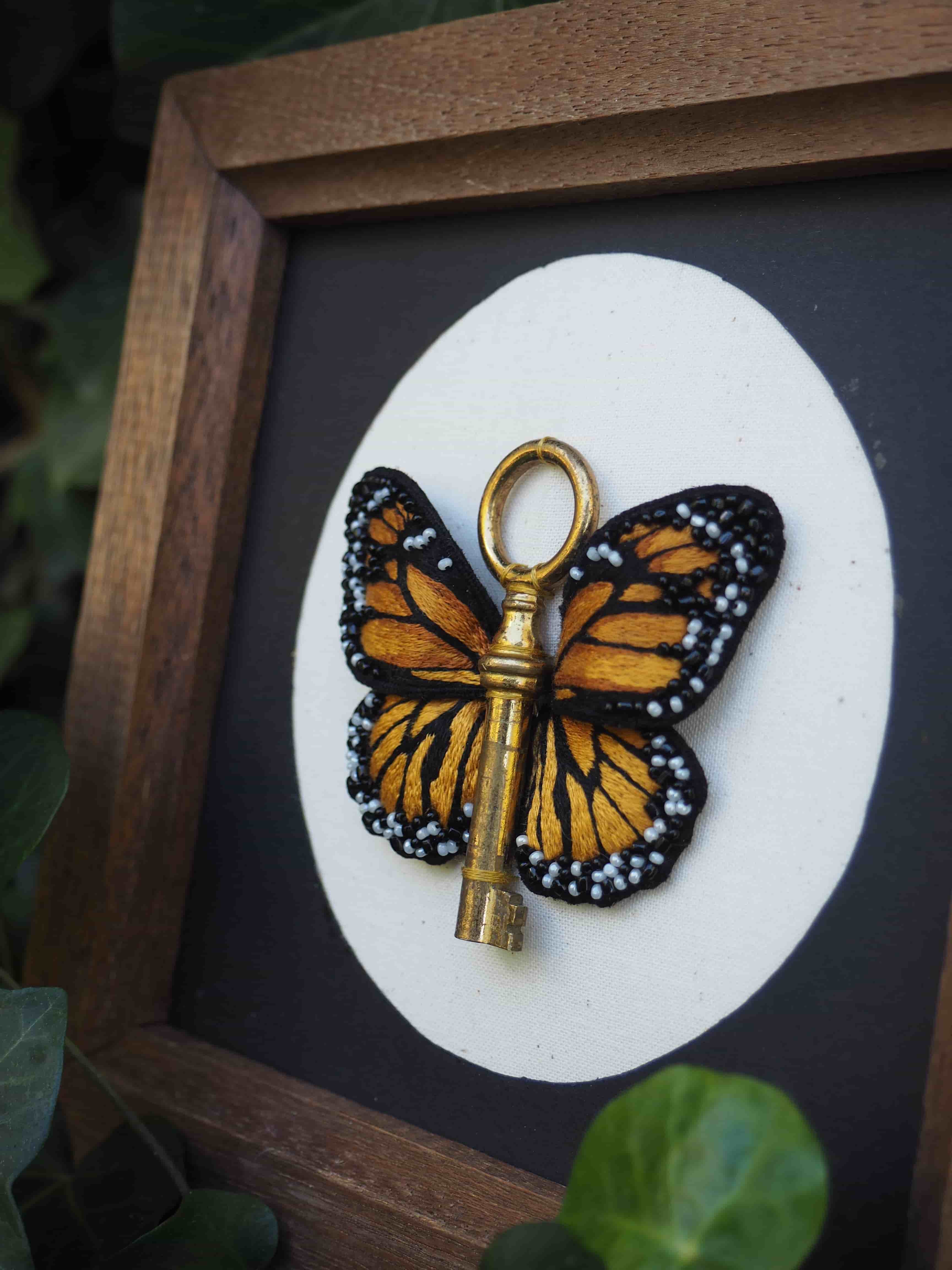 Cette image présente une œuvre d'art encadrée très créative et symbolique. Voici sa description détaillée : **Composition principale :**
- Un cadre en bois naturel de forme carrée
- Un fond noir mat qui fait ressortir l'élément central
- Un cercle blanc au centre servant de support à l'œuvre **Élément artistique central :**
- Une clé dorée/bronze transformée artistiquement
- Les ailes d'un papillon monarque (orange vif avec bordures noires et points blancs) ont été ajoutées à la clé
- La tête de la clé forme le "corps" du papillon
- L'anneau de la clé sert d'élément de suspension décoratif **Symbolisme :**
Cette création combine deux symboles puissants :
- La clé : représentant l'accès, les solutions, les opportunités
- Le papillon monarque : symbolisant la transformation, la liberté, la métamorphose **Contexte :**
L'œuvre est photographiée sur ce qui semble être une surface naturelle avec de la végétation visible en arrière-plan, suggérant un environnement extérieur ou près d'une fenêtre. Cette pièce artistique unique mélange habilement l'utilitaire et le naturel pour créer une métaphore visuelle sur la transformation et les nouvelles possibilités.