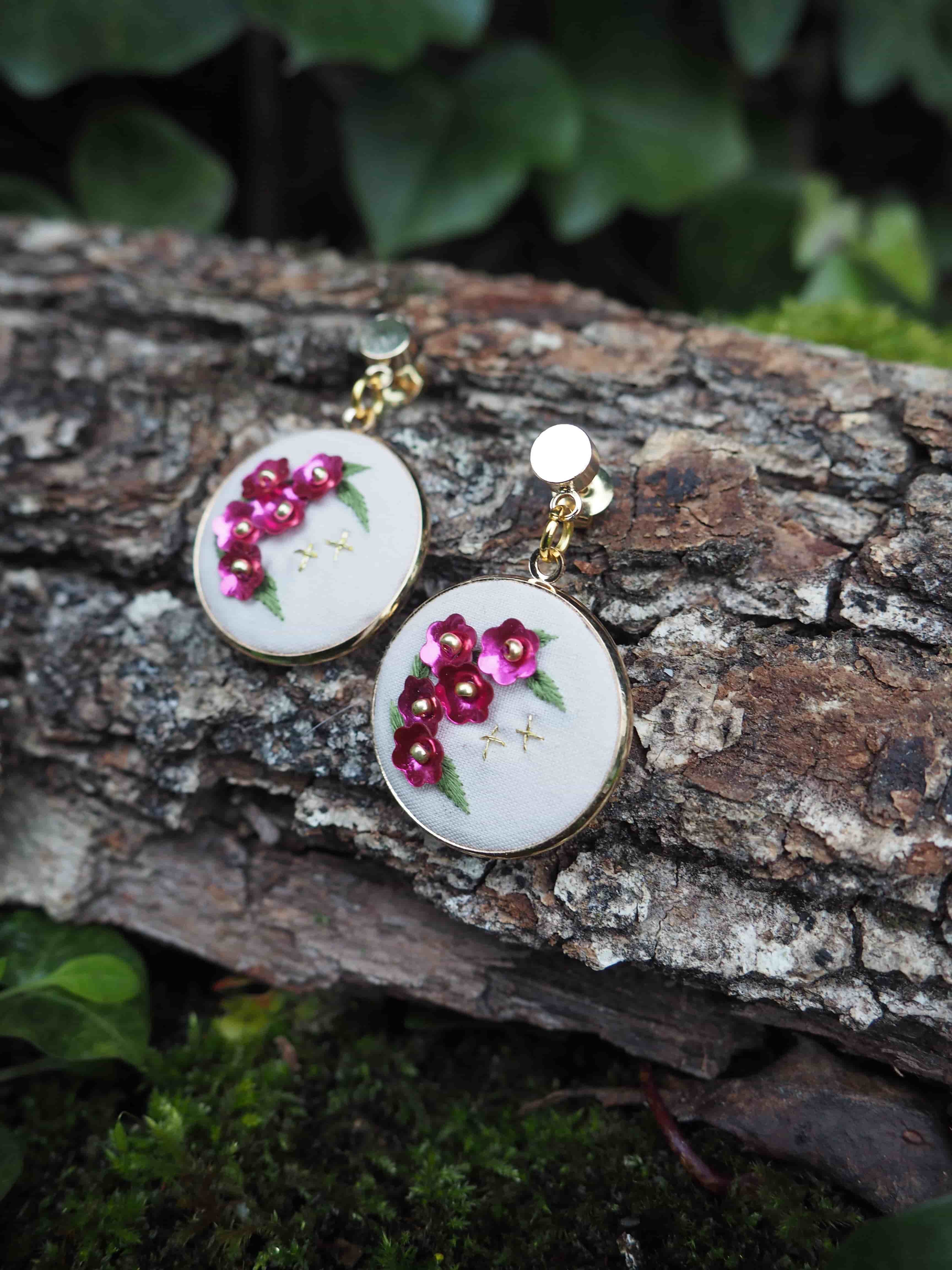 L'image montre une paire de boucles d'oreilles artisanales posées sur un morceau d'écorce d'arbre. Ces boucles d'oreilles sont de forme ronde et semblent être faites de tissu blanc brodé. Sur chaque boucle d'oreille, on peut voir de délicates fleurs brodées en rose foncé, probablement des roses ou des fleurs similaires. Les boucles d'oreilles ont une apparence très délicate et féminine. Elles sont attachées à des tiges dorées, ce qui suggère qu'elles sont conçues pour être portées aux oreilles. Le contraste entre la texture rugueuse de l'écorce et la finesse des broderies est frappant. L'arrière-plan montre un environnement naturel flou, avec des feuilles vertes visibles, ce qui donne à l'image une ambiance forestière ou de jardin. Cette mise en scène met en valeur la beauté naturelle et artisanale des bijoux. Cette photographie semble mettre l'accent sur la délicatesse du travail manuel et l'harmonie entre les créations artisanales et la nature.