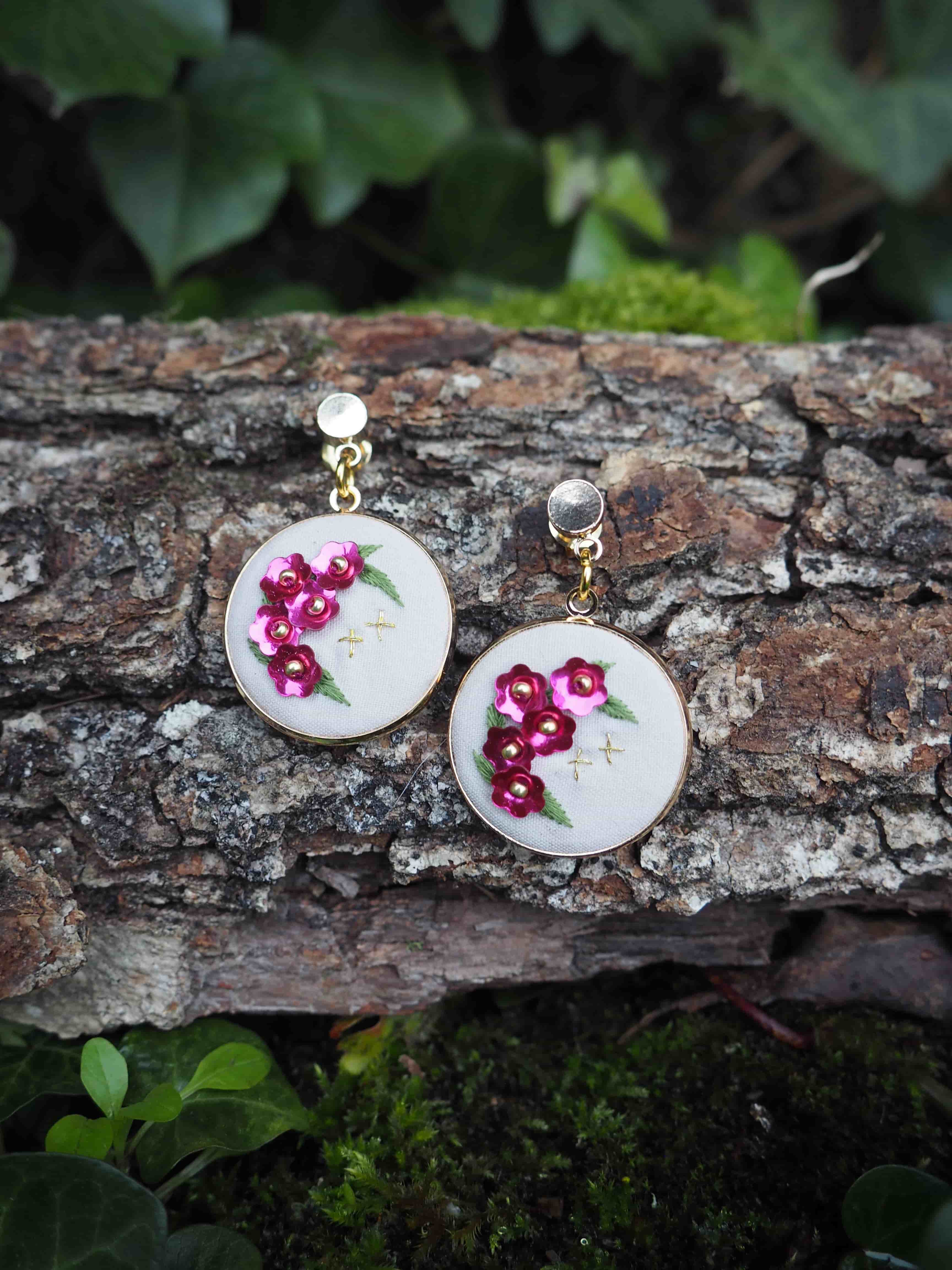 L'image montre une paire de boucles d'oreilles artisanales posées sur un morceau d'écorce d'arbre. Les boucles d'oreilles sont constituées de petits cercles blancs sur lesquels sont brodées des fleurs roses. Ces fleurs semblent être des cerisiers en fleurs ou des fleurs de pommier, brodées avec du fil rose vif. Les pendentifs sont attachés à des tiges dorées, probablement en métal. L'arrière-plan montre l'écorce rugueuse et texturée de l'arbre, avec quelques feuilles vertes floues visibles autour des bords de l'image, suggérant un cadre naturel en plein air. Cette photographie met en valeur le contraste entre les délicates boucles d'oreilles faites à la main et la texture brute de l'écorce, créant une composition intéressante qui mêle artisanat et nature. L'image semble capturer un moment de présentation créative de bijoux artisanaux dans un environnement naturel.
