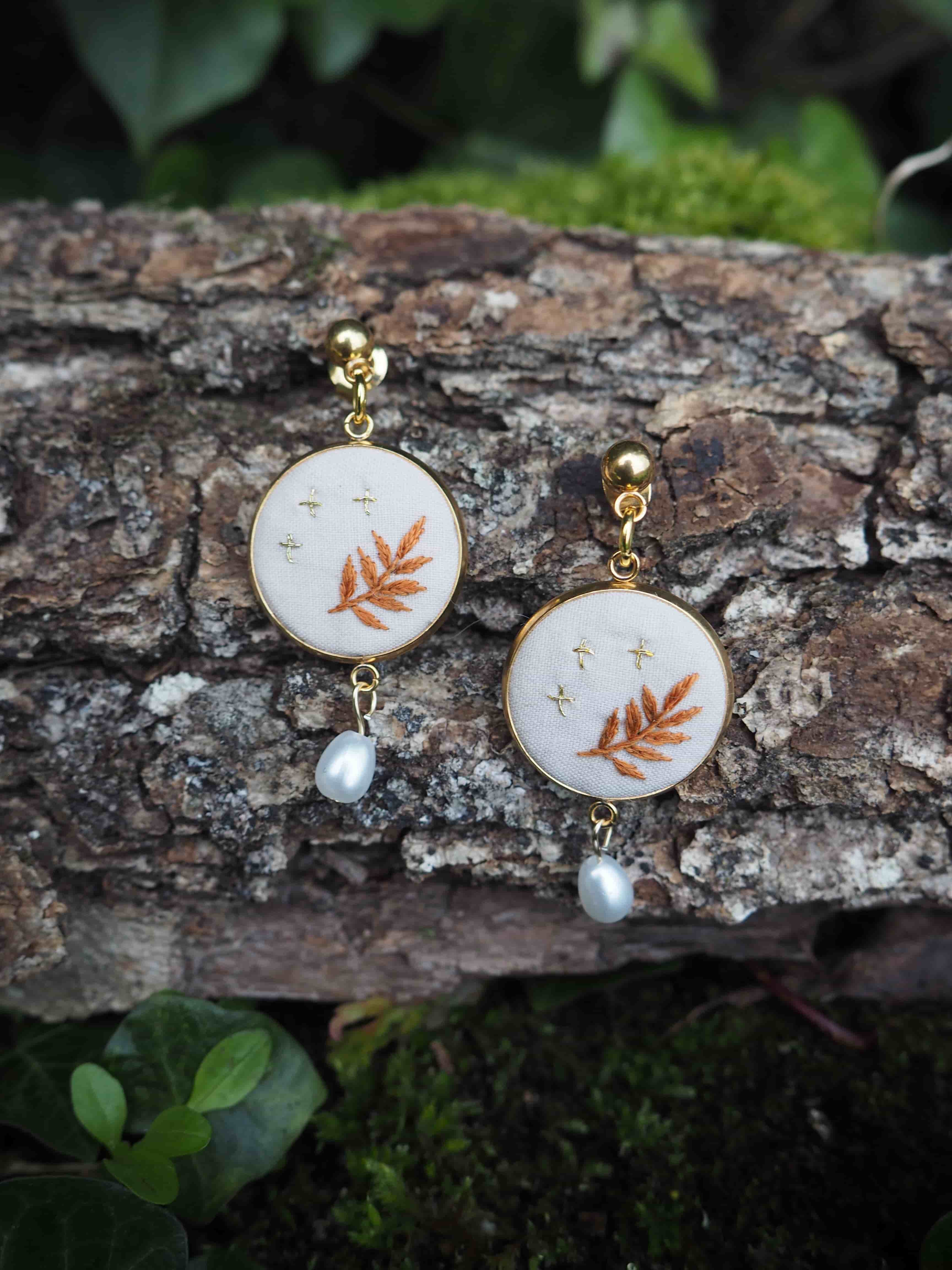 L'image montre une paire de boucles d'oreilles délicates posées sur un morceau d'écorce d'arbre. Les boucles d'oreilles sont composées de petits cercles blancs sur lesquels est brodé un motif de feuille orange ou cuivré. Sous chaque cercle pend une petite perle blanche. Les boucles d'oreilles semblent être faites à la main, avec un design naturel et organique qui s'harmonise bien avec le fond d'écorce rugueuse. L'arrière-plan montre également quelques feuilles vertes floues, suggérant un cadre naturel en extérieur. Le contraste entre la délicatesse des bijoux et la texture rugueuse de l'écorce crée une composition intéressante. Les couleurs douces des boucles d'oreilles - le blanc, l'orange pâle et l'éclat des perles - se détachent joliment sur les tons bruns de l'écorce. Cette image pourrait être utilisée pour présenter des bijoux artisanaux ou pour illustrer un style de mode inspiré par la nature.