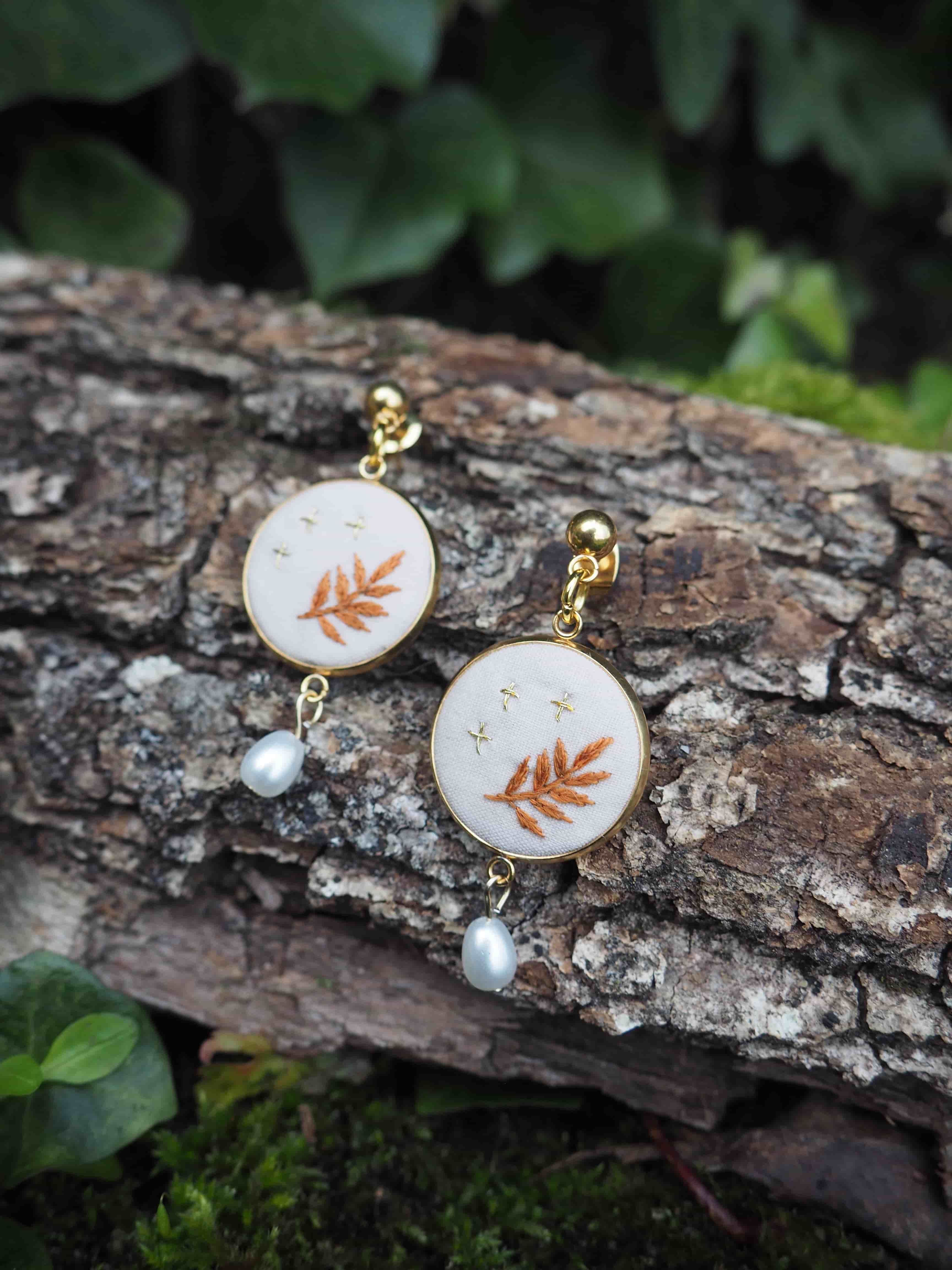 L'image montre une paire de boucles d'oreilles délicates posées sur un morceau de bois ou d'écorce. Les boucles d'oreilles sont composées de petits médaillons circulaires blancs, chacun orné d'une feuille de fougère de couleur cuivrée ou dorée. Sous chaque médaillon pend une petite perle blanche. Les boucles d'oreilles semblent être faites d'or ou d'un métal doré, ce qui contraste joliment avec le fond naturel rugueux de l'écorce. L'arrière-plan montre des feuilles vertes floues, suggérant que la photo a été prise dans un environnement naturel, peut-être un jardin ou une forêt. Cette image met en valeur la beauté délicate des bijoux inspirés par la nature, en les plaçant dans un cadre naturel qui complimente leur design. Le contraste entre les bijoux raffinés et la texture brute de l'écorce crée une composition intéressante et attrayante.