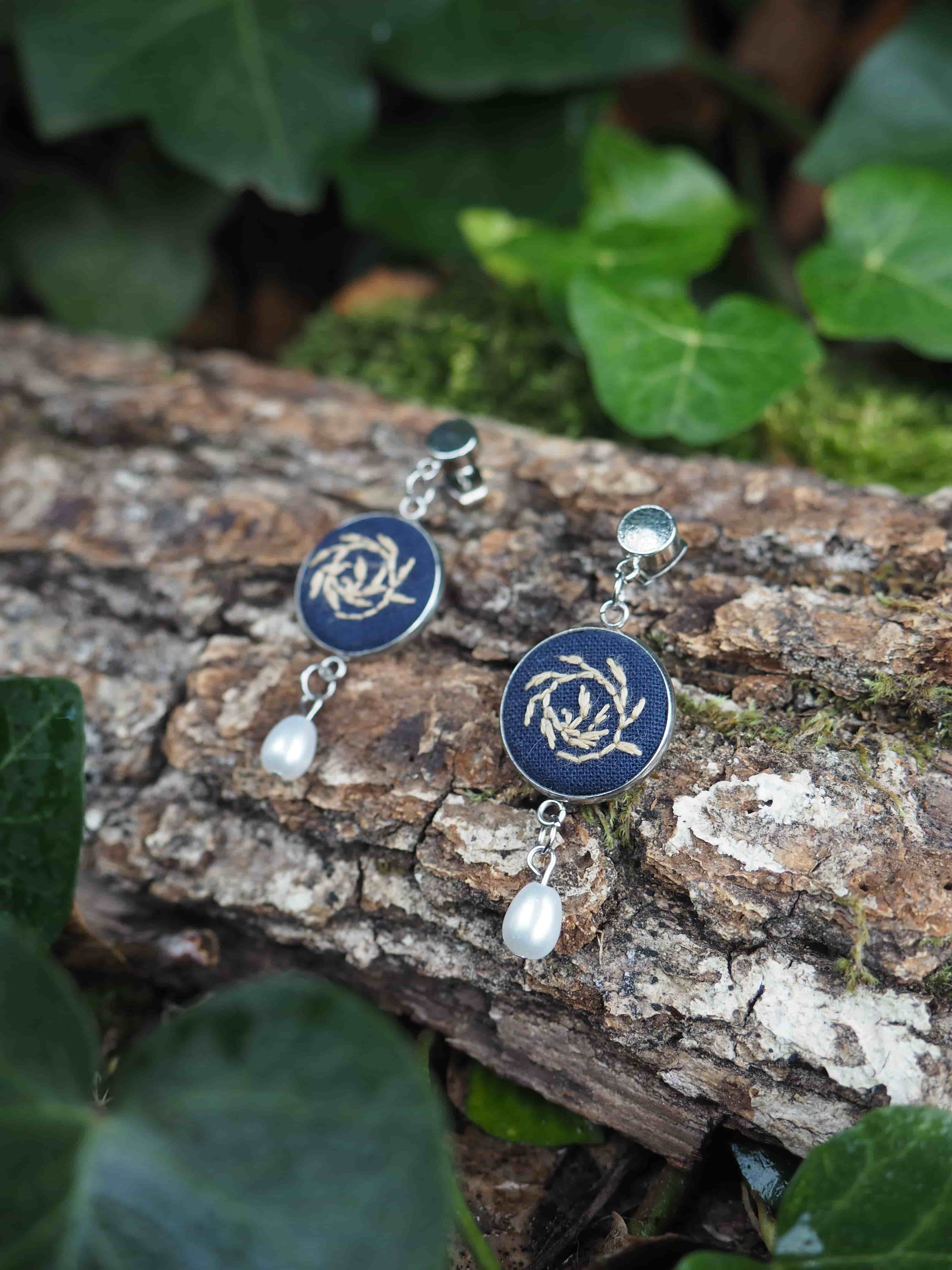 Cette image présente une paire de boucles d'oreilles élégantes disposées sur une branche d'arbre dans un environnement naturel.  **Description des bijoux :**
- Boucles d'oreilles pendantes avec des éléments circulaires bleu marine ornés de motifs décoratifs argentés en spirale ou entrelacs
- Perles blanches nacrées suspendues en pendentif
- Attaches et connecteurs en métal argenté
- Style artisanal avec une esthétique à la fois moderne et intemporelle **Mise en scène :**
- Les bijoux reposent sur une branche d'arbre rugueuse et texturée
- Arrière-plan composé de feuillage vert luxuriant légèrement flouté
- Éclairage naturel qui met en valeur les détails et les couleurs
- Composition photographique soignée créant un contraste harmonieux entre les éléments manufacturés et l'environnement naturel Cette présentation met particulièrement en valeur le caractère artisanal des bijoux tout en créant une atmosphère organique et apaisante.