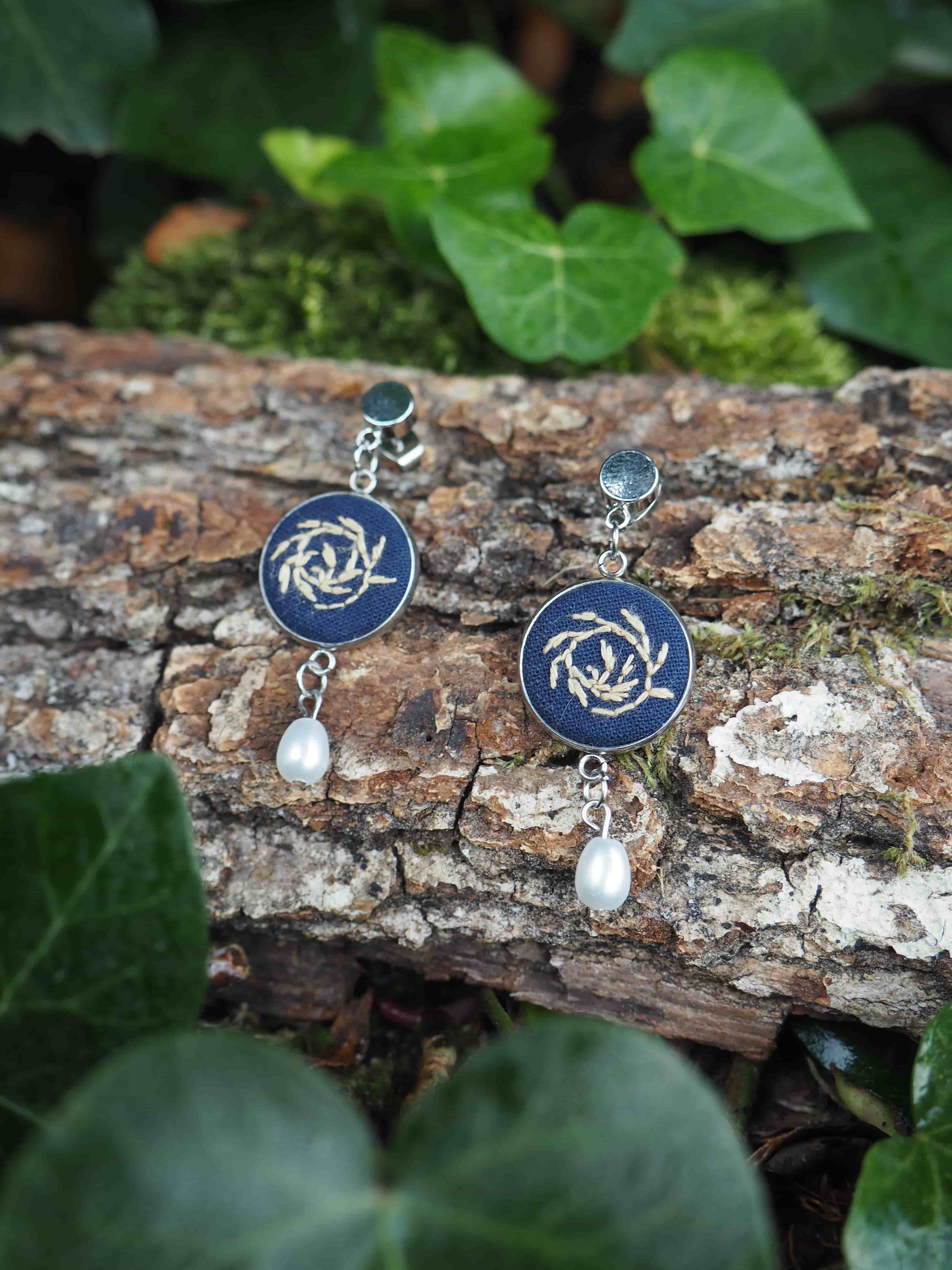 Cette image présente une paire de boucles d'oreilles élégantes photographiées dans un cadre naturel. Voici les détails que je peux observer : **Les boucles d'oreilles :**
- Elles comportent des pendentifs circulaires de couleur bleu foncé/marine
- Chaque pendentif est orné d'un motif décoratif doré ou argenté au centre, qui ressemble à un design floral ou géométrique stylisé
- Des perles blanches nacrées pendent sous chaque cercle bleu
- Le système d'attache semble être en métal argenté **Le cadre naturel :**
- Les bijoux sont posés sur un morceau d'écorce d'arbre ou de bois brut, avec une texture rugueuse et des tons bruns naturels
- L'arrière-plan est composé de feuillage vert luxuriant, légèrement flou, créant un effet de profondeur de champ
- On distingue de la mousse verte sur l'écorce
- L'éclairage naturel met bien en valeur les couleurs et les détails des bijoux Cette mise en scène crée un contraste harmonieux entre l'élégance des bijoux et la beauté brute de la nature environnante.