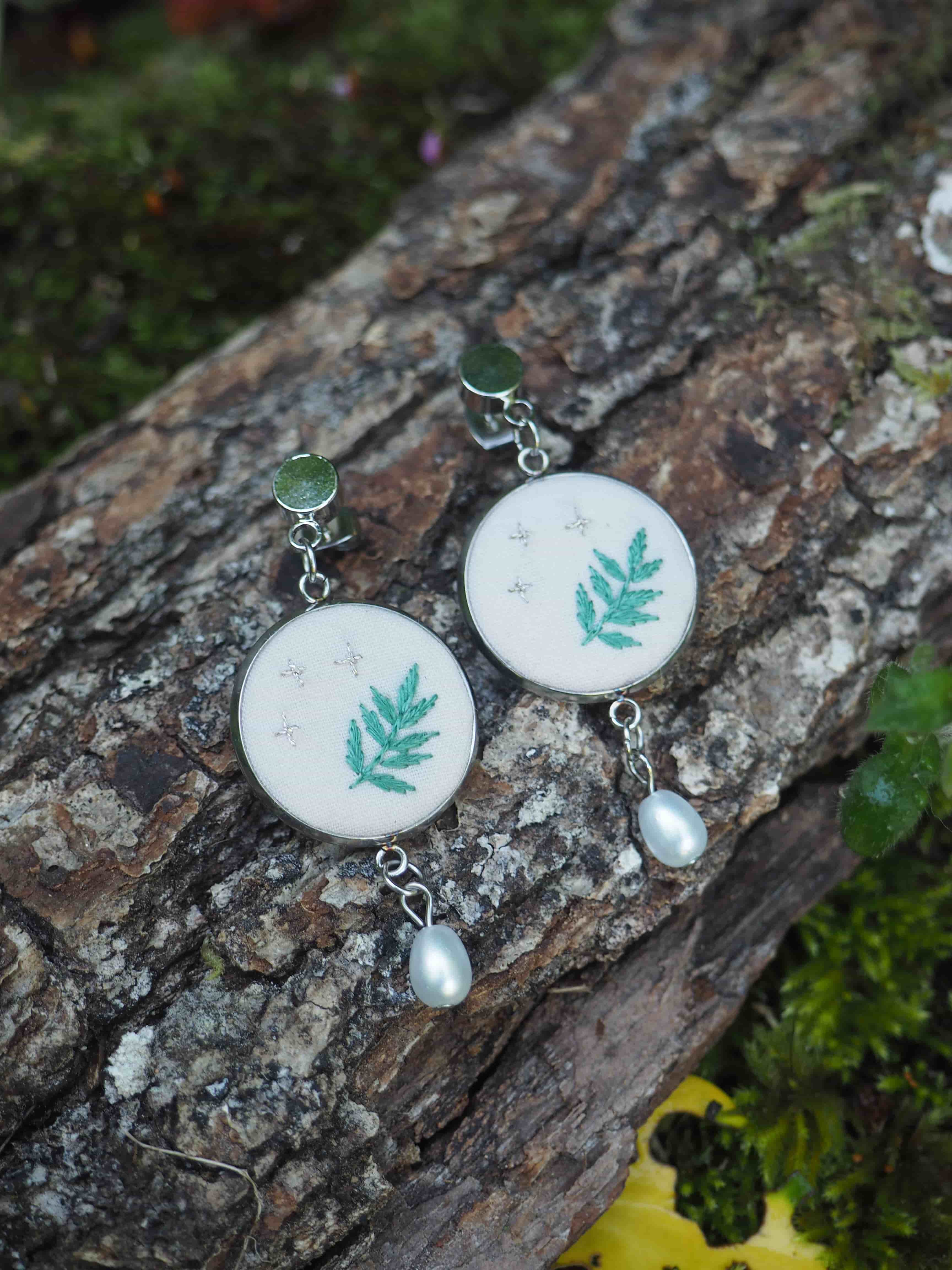 Ces boucles d'oreilles sont de délicates créations artisanales qui présentent un motif de feuille turquoise brodé ou imprimé sur un fond blanc circulaire. Chaque boucle d'oreille est ornée d'une perle nacrée suspendue au bas du médaillon principal. Les attaches semblent être en métal argenté avec de petites pierres ou cabochons verts. L'image montre les bijoux posés sur ce qui semble être un morceau de bois ou d'écorce texturé, avec de la mousse verte visible en arrière-plan, créant une ambiance naturelle et forestière qui complimente parfaitement le design botanique des boucles d'oreilles.