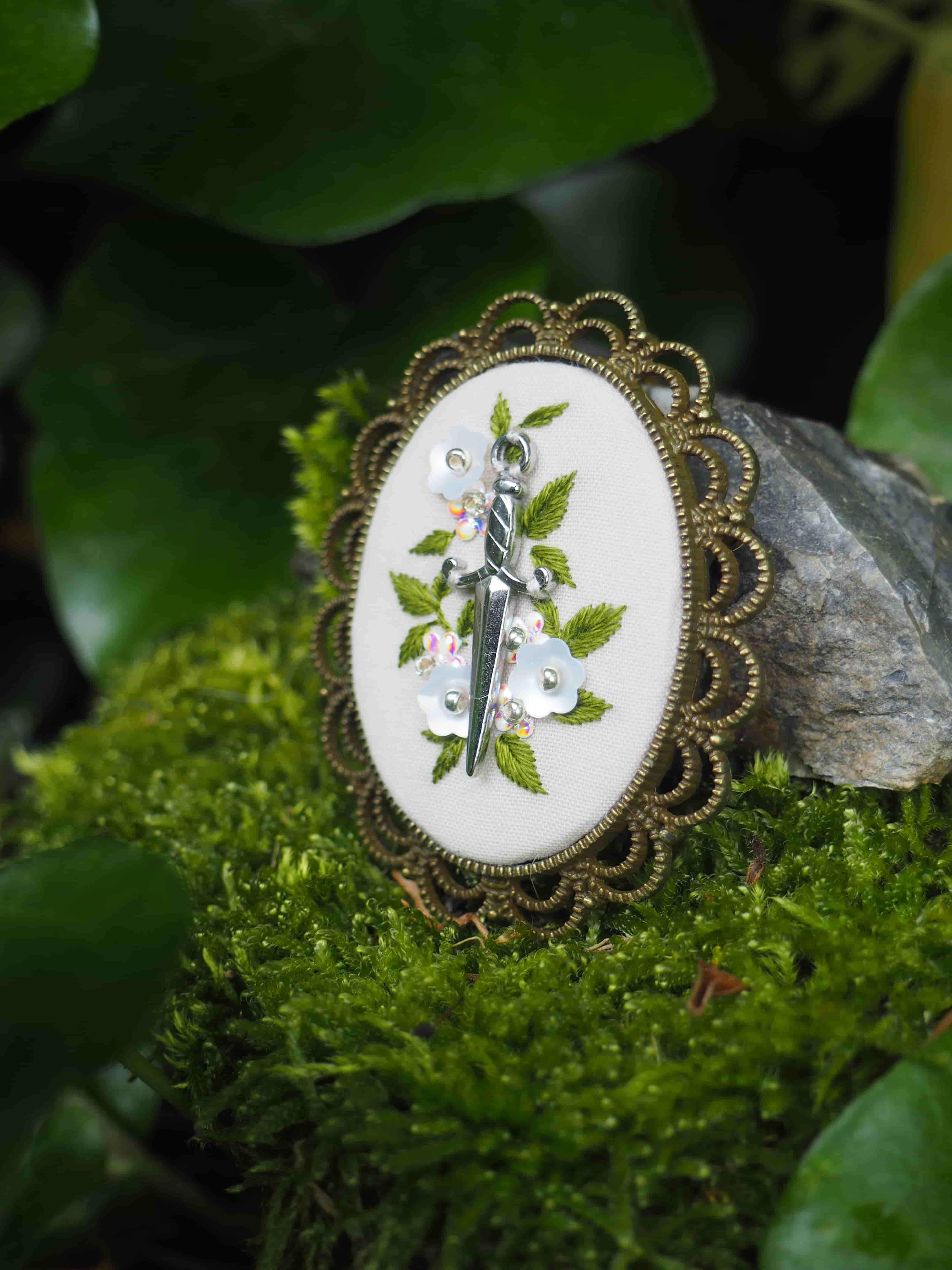 L'image montre trois broches ou bijoux décoratifs vintage disposés sur de la mousse verte. Ces broches semblent être de style victorien ou ancien, avec des cadres en métal ornementés de motifs floraux. Chaque broche contient ce qui semble être une peinture ou une illustration sur fond blanc représentant des motifs de feuilles vertes. Les broches sont photographiées dans un cadre naturel, posées sur de la mousse verte luxuriante, ce qui crée un contraste intéressant entre les éléments manufacturés et naturels. L'éclairage et la composition de la photo créent une ambiance féerique et romantique.