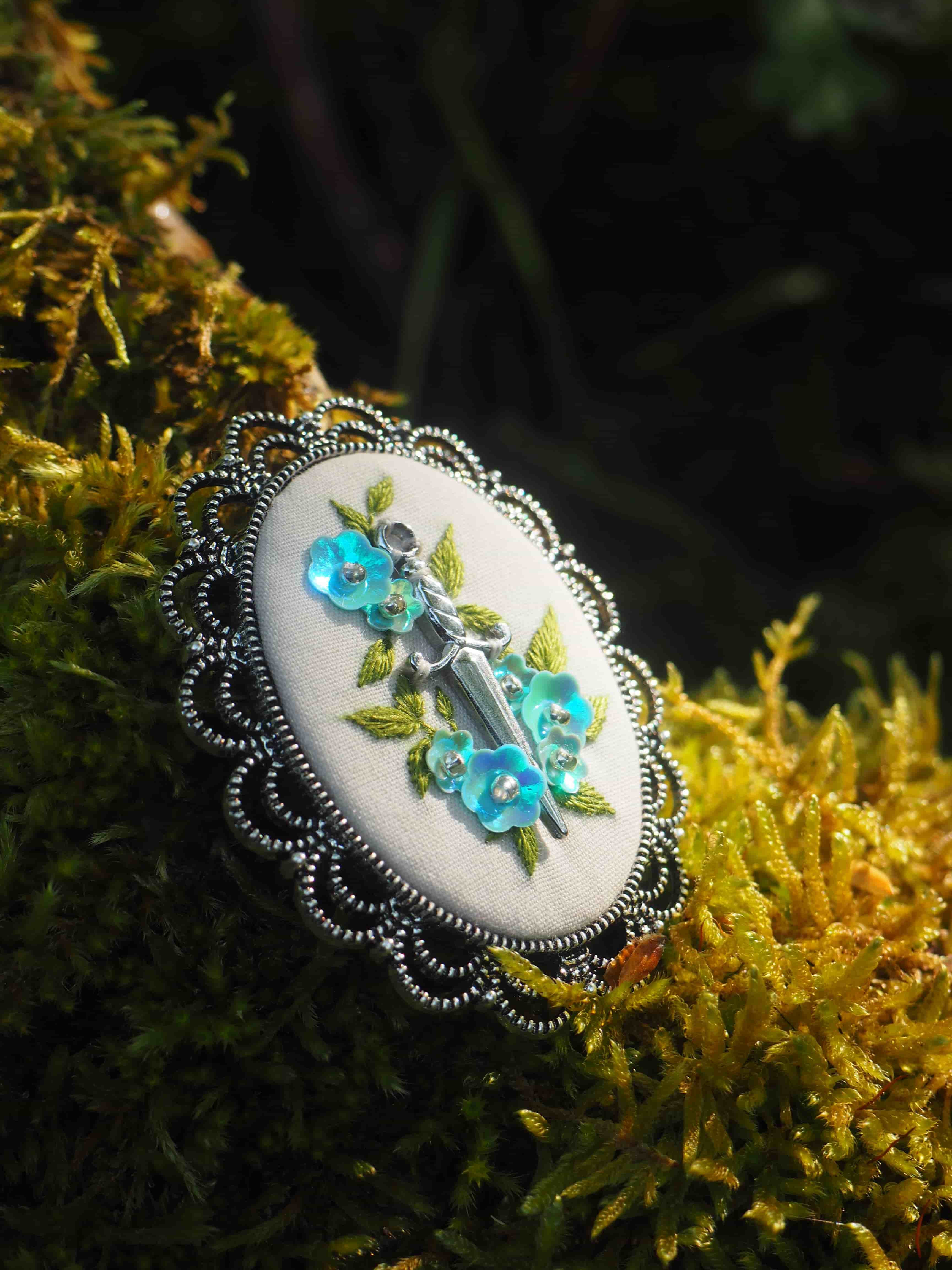 L'image montre un bijou ou une broche décorative posée sur ce qui semble être de la mousse ou une plante jaune-dorée. Le bijou a une forme ovale avec un bord décoratif en métal argenté au motif ajouré. Le centre est de couleur blanche ou crème et comporte une décoration florale avec des petites fleurs bleues et des feuilles vertes qui semblent être peintes ou brodées. La pièce a un aspect vintage ou artisanal, avec un style romantique et délicat. Le contraste entre le bijou clair et le fond végétal doré crée une belle composition photographique.