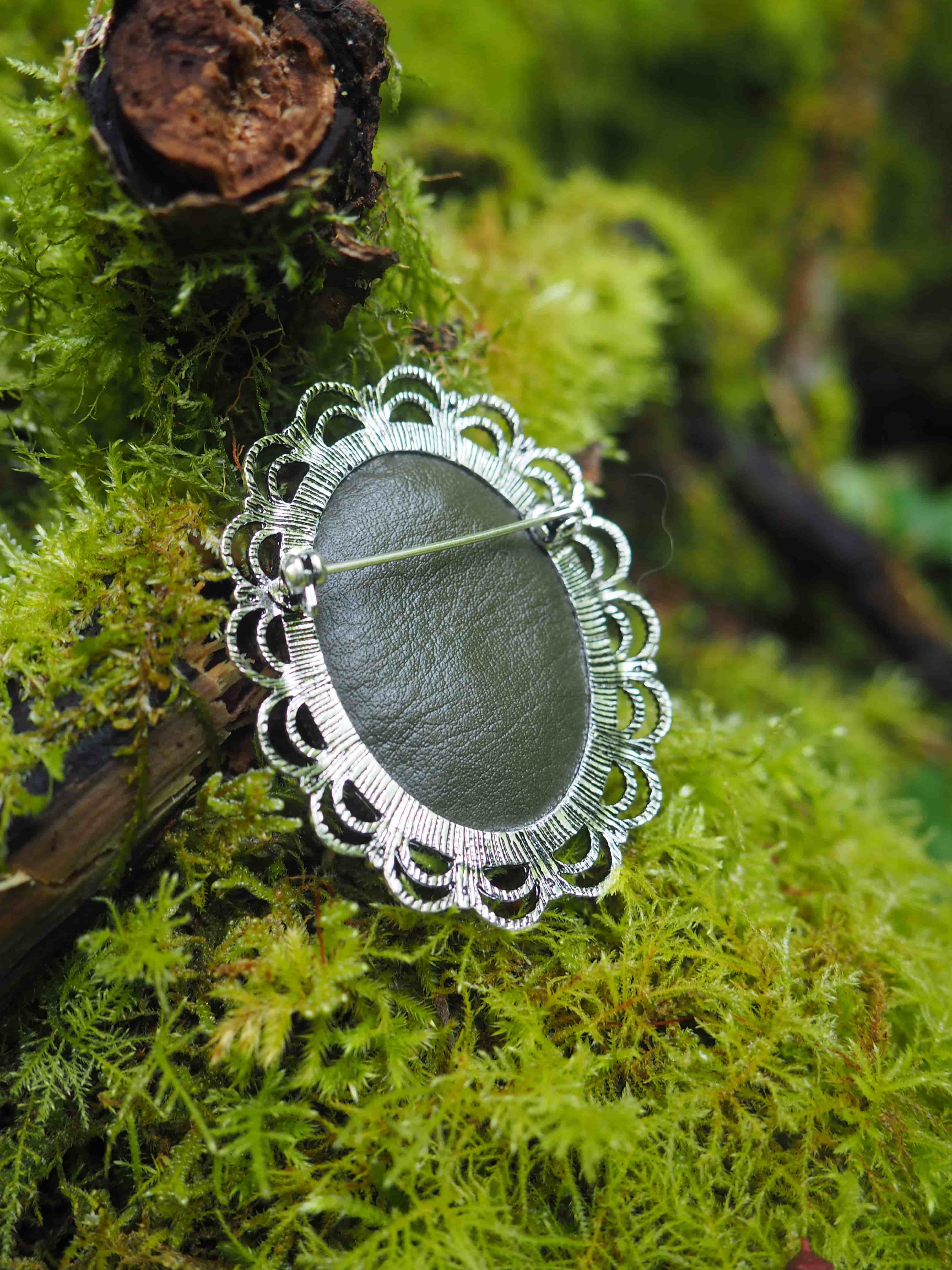 L'image montre des bijoux ou des ornements décoratifs posés sur un lit de mousse verte luxuriante. Ces ornements semblent être des broches ou des pendentifs avec un centre foncé (probablement en cuir ou en pierre) entouré d'une bordure en dentelle blanche délicate. La dentelle forme un motif décoratif rayonnant qui rappelle une fleur ou un soleil. Le contraste entre le vert vif de la mousse et le blanc de la dentelle crée une composition très esthétique et naturelle. On peut également apercevoir quelques morceaux d'écorce ou de bois foncé en arrière-plan.