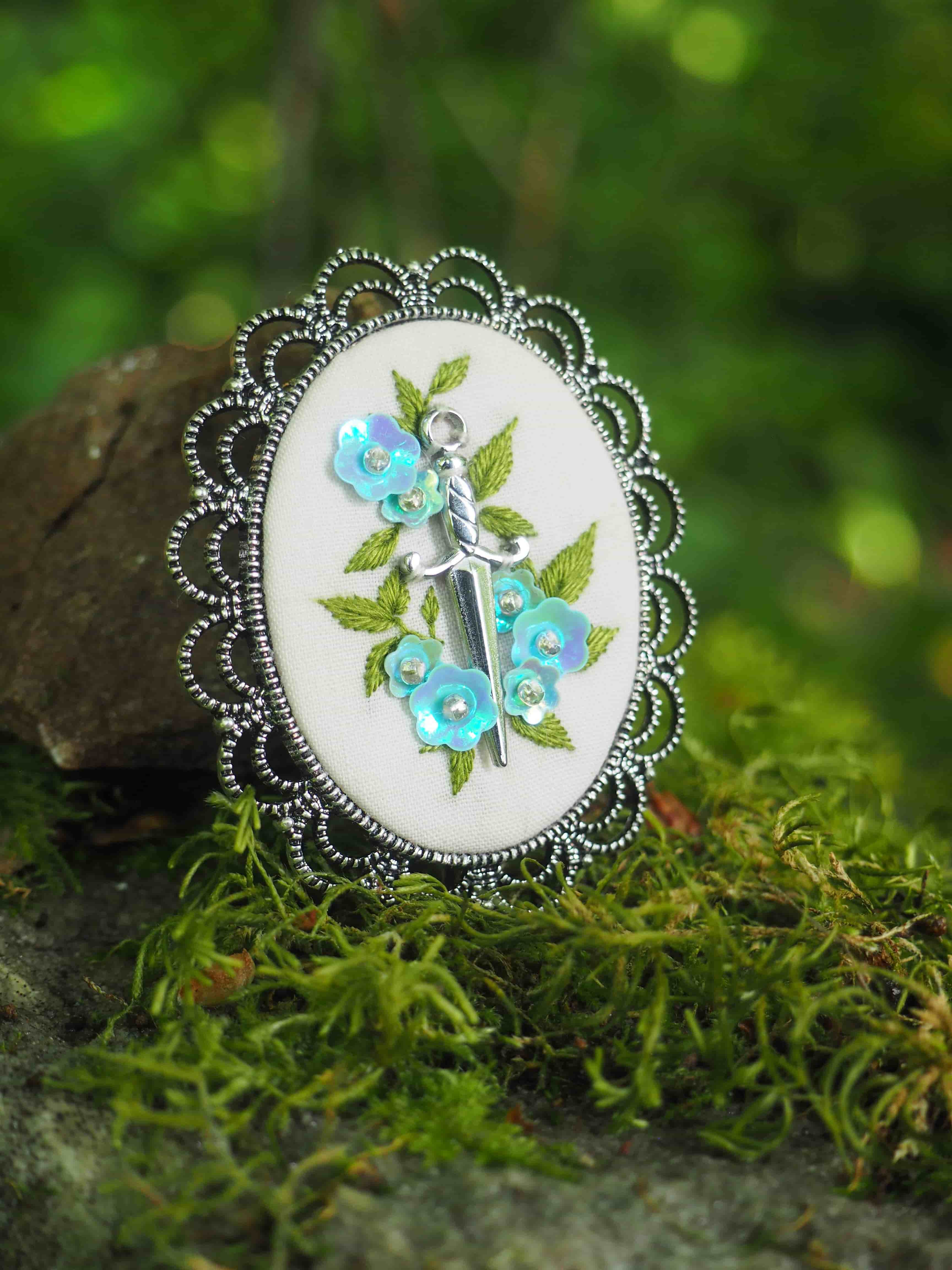 Cette image montre une belle broche ou un pendentif décoratif avec un cadre en métal argenté au motif festonné. Le centre présente une broderie délicate sur un fond blanc, représentant des fleurs bleues avec des feuilles vertes. Le design est élégant et délicat, avec un style vintage ou romantique. La photo est prise sur un fond naturel de mousse verte, ce qui crée un beau contraste avec le bijou et lui donne une ambiance féerique ou enchantée. L'éclairage doux et le cadre naturel mettent parfaitement en valeur les détails délicats de cette pièce artisanale.