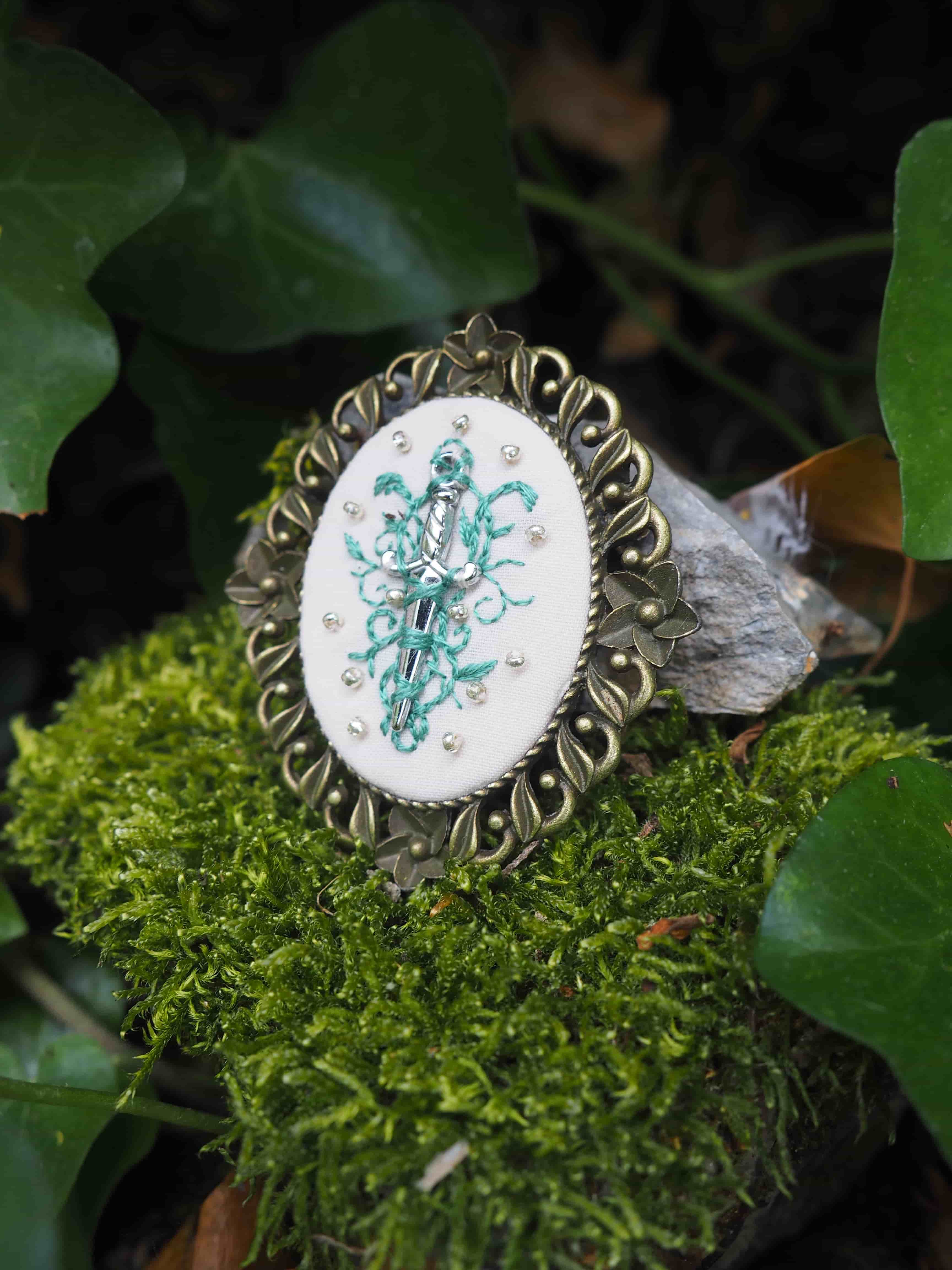 Cette image montre un joli bijou ou broche vintage posé sur de la mousse verte dans un cadre naturel. Le bijou présente un médaillon ovale avec un motif floral ou décoratif de couleur turquoise sur fond blanc, entouré d'un cadre ornementé en métal de style antique avec des motifs décoratifs. L'arrière-plan montre quelques feuilles vertes et de la mousse, donnant à la photo une ambiance naturelle et féerique. La composition est très artistique, mêlant l'aspect délicat du bijou avec les éléments naturels qui l'entourent.