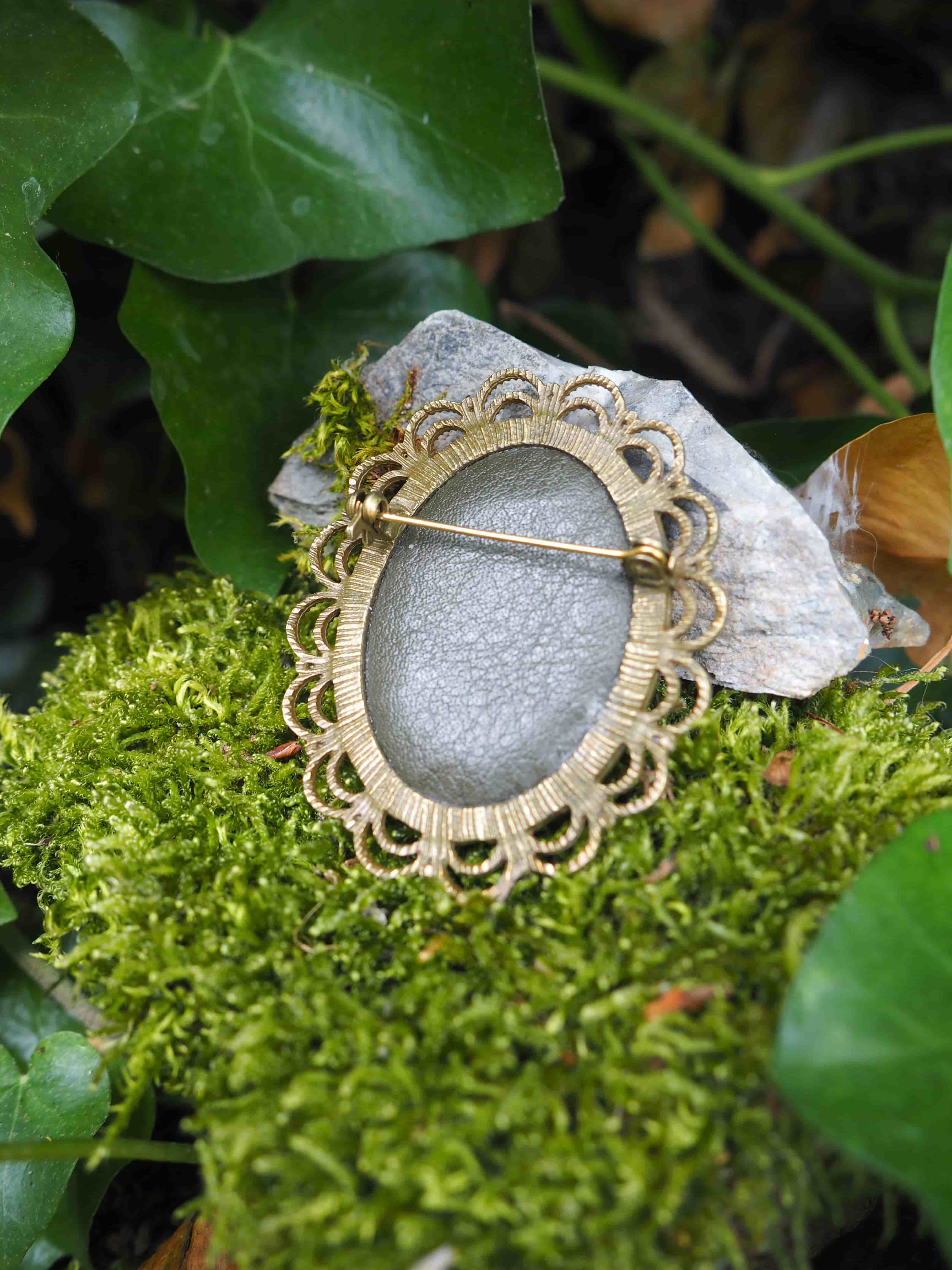 L'image montre des bijoux ou des broches vintage posés sur un lit de mousse verte. Les pièces semblent être des broches ou des pendentifs de style ancien avec des motifs décoratifs ornementaux autour de ce qui paraît être des pierres ou des cabochons gris/argentés au centre. Le cadre naturel avec la mousse verte et quelques feuilles en arrière-plan crée une ambiance féerique et romantique. Le contraste entre les éléments métalliques dorés des bijoux et la texture naturelle de la mousse verte est particulièrement esthétique.