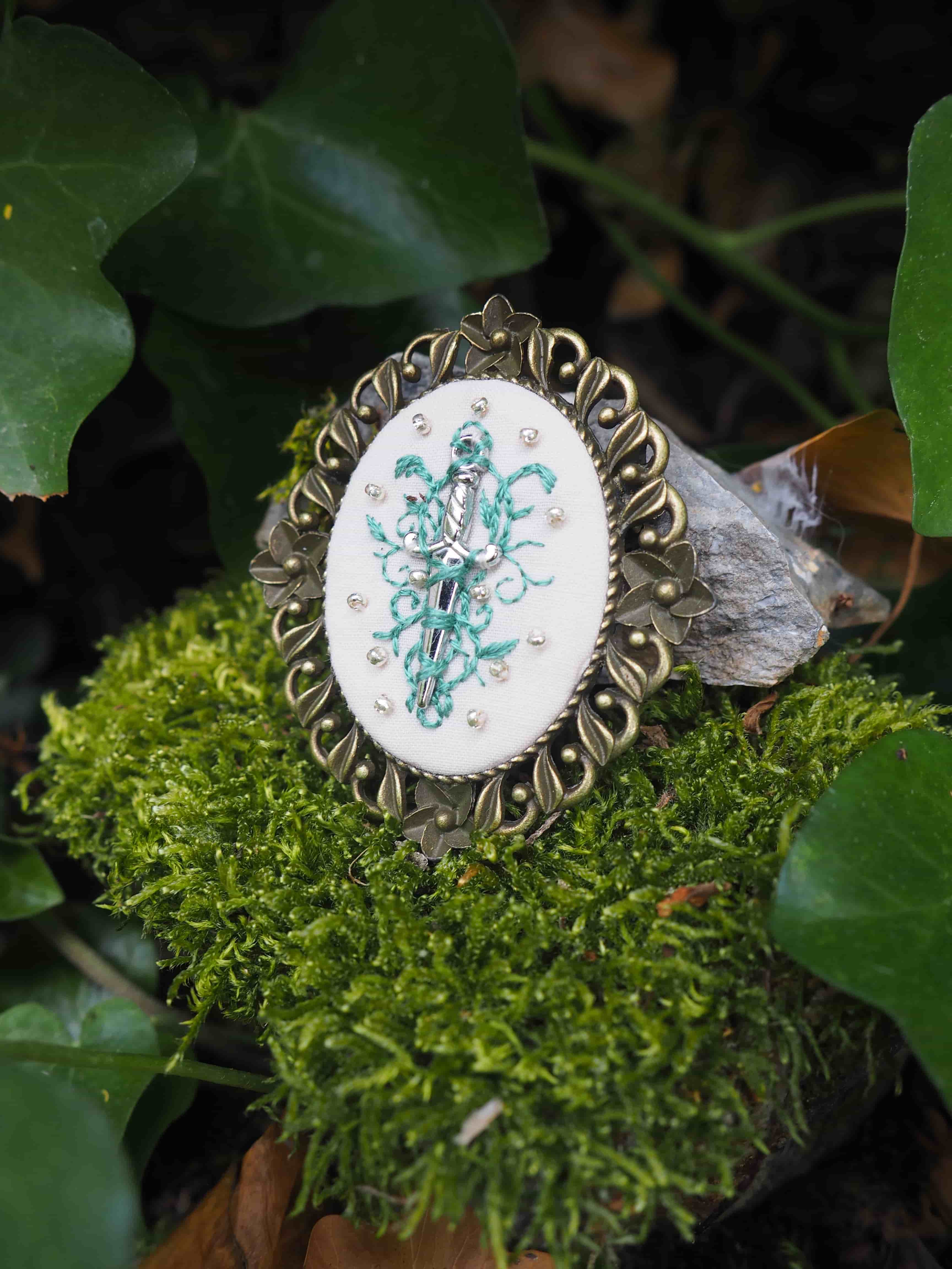 Cette image montre un joli bijou ou broche vintage posé sur un lit de mousse verte. Le bijou a un cadre décoratif en métal de style baroque ou victorien avec des motifs ornementaux. Au centre se trouve un médaillon rond avec une délicate broderie ou un dessin floral en turquoise sur fond blanc. L'ensemble est photographié dans un cadre naturel avec des feuilles vertes en arrière-plan, créant un contraste intéressant entre l'objet artisanal et l'environnement naturel. La composition est artistique et évoque une ambiance féerique ou romantique.