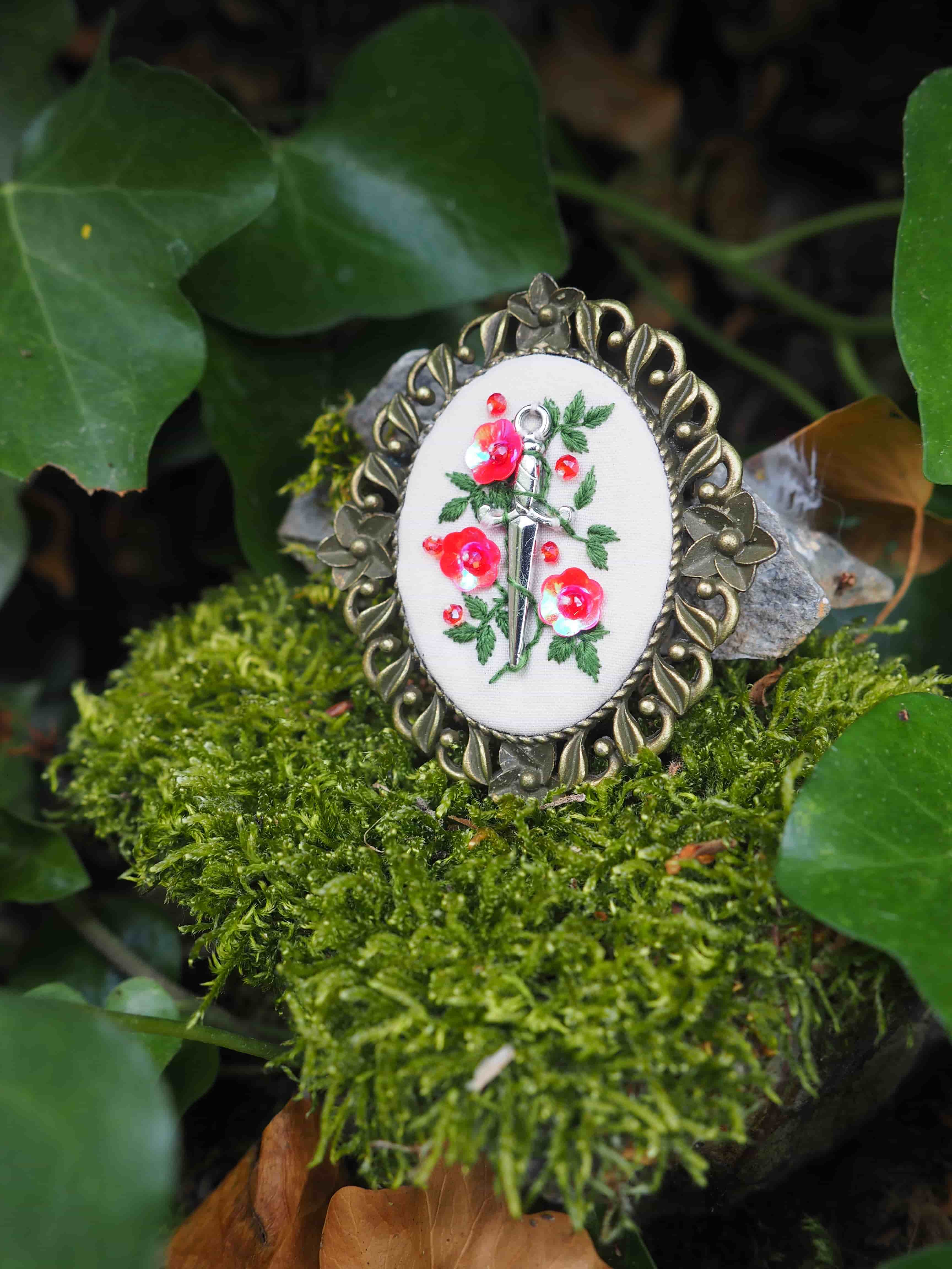 Cette image montre une broche ou un médaillon décoratif posé sur de la mousse verte. Le bijou a un cadre en métal de style vintage avec des motifs ornementaux ajourés. Au centre se trouve une pièce brodée ou peinte représentant des roses rouges et un motif de dague ou d'épée sur un fond blanc. Les détails sont délicats et le contraste entre le rouge des fleurs et le fond blanc est très net. L'arrière-plan naturel avec la mousse verte et quelques feuilles crée une mise en scène romantique et bucolique pour ce bijou artisanal.