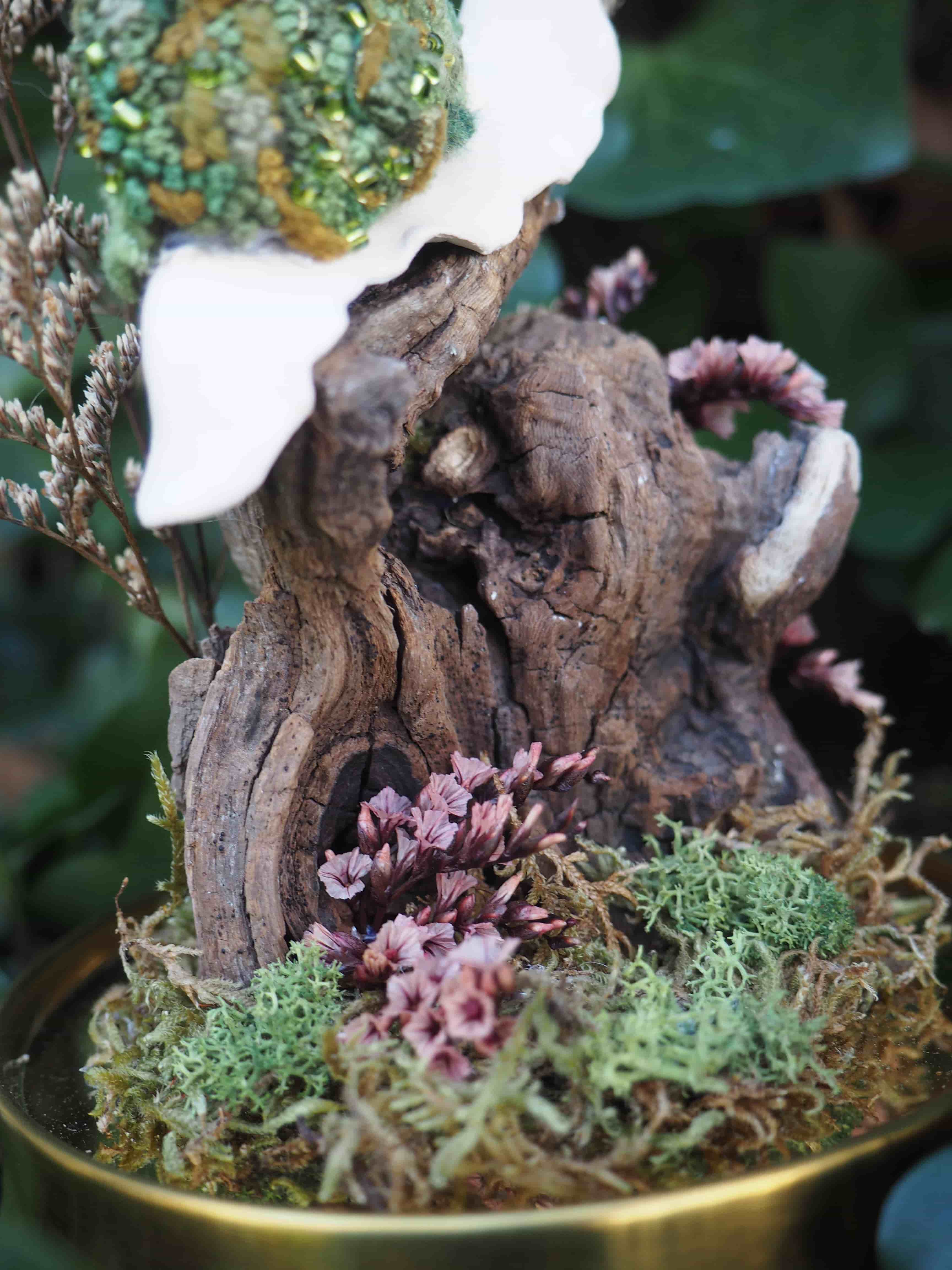 Cette image présente une composition miniature et détaillée, évoquant un petit monde féerique ou une scène de forêt enchantée. Au centre de l'image, on voit un morceau de bois flotté ou de souche d'arbre, de couleur marron foncé avec des textures et des creux naturels. Il semble servir de base ou de structure principale à la composition. Autour de ce bois, et sur une base qui pourrait être un plateau doré ou en laiton (dont on aperçoit le bord inférieur), se trouve un tapis de mousse verte de différentes nuances, créant un effet de sous-bois. Des petites fleurs ou plantes séchées de couleur rose-mauve sont parsemées, ajoutant des touches de couleur et de délicatesse. En haut de la composition, une forme blanche et lisse, qui pourrait être une feuille stylisée, une aile ou un élément décoratif en céramique, s'élève du bois. Sur cette forme blanche, ou juste au-dessus, repose un objet sphérique ou ovoïde, texturé et de couleur verte avec des reflets dorés ou brillants, rappelant une pierre précieuse, une gemme ou un élément végétal orné. L'arrière-plan est flou, mais on distingue des feuillages verts, suggérant un environnement naturel ou un jardin. L'ensemble de l'image dégage une atmosphère de calme, de mystère et de beauté miniature, avec une attention particulière aux détails et aux textures.