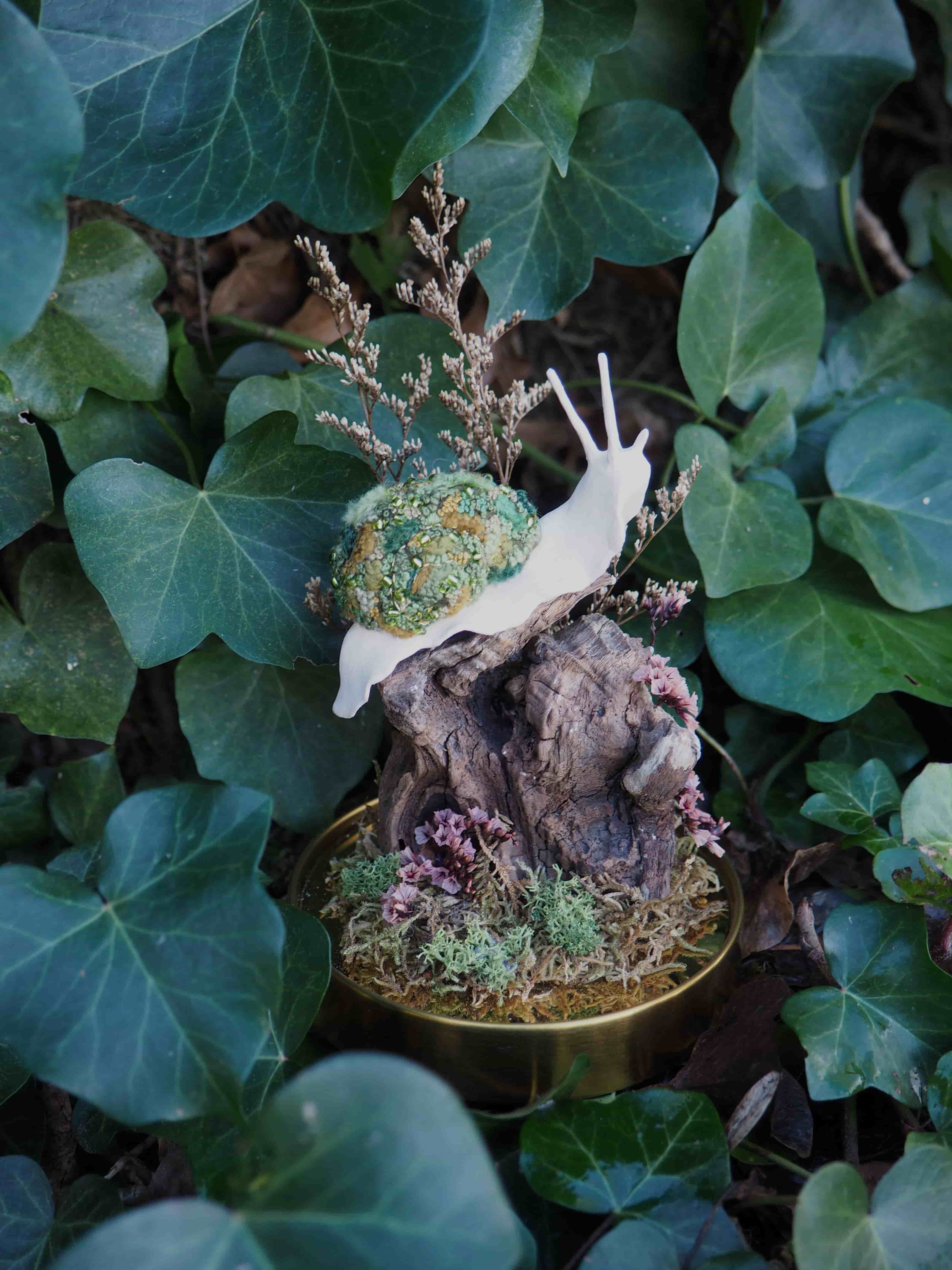 L'image présente une figurine d'escargot blanc avec une coquille texturée verte et dorée, posée sur un petit socle décoratif. **Détails de la figurine :**
*   **Escargot :** Le corps de l'escargot est d'un blanc uni et lisse, avec des antennes fines et allongées.
*   **Coquille :** La coquille est la partie la plus distinctive, recouverte d'une texture granuleuse et scintillante, mélangeant des tons de vert clair, de vert foncé et de doré. Elle semble être faite de petites perles ou de paillettes.
*   **Socle :** L'escargot est perché sur un petit morceau de bois ou de roche de couleur marron foncé, qui sert de base. Autour de ce support, on observe de la mousse verte et des petites touffes de végétation rose ou violette, créant un mini-écosystème.
*   **Décoration florale :** Derrière l'escargot, une petite branche de fleurs séchées ou de feuillage fin de couleur beige-brun ajoute une touche délicate à la composition. **Contexte et arrière-plan :**
La figurine est placée au milieu d'un feuillage dense de lierre vert foncé, qui forme un arrière-plan naturel et luxuriant. Les feuilles de lierre sont de différentes tailles et teintes de vert, certaines étant plus claires et d'autres plus sombres, créant de la profondeur. La lumière semble douce et diffuse, mettant en valeur les détails de la figurine sans créer d'ombres dures. **Ambiance générale :**
L'image dégage une atmosphère féerique et délicate, rappelant un jardin miniature ou un conte de fées. La figurine d'escargot, avec sa coquille scintillante, est le point focal et attire l'attention par son originalité et sa beauté.