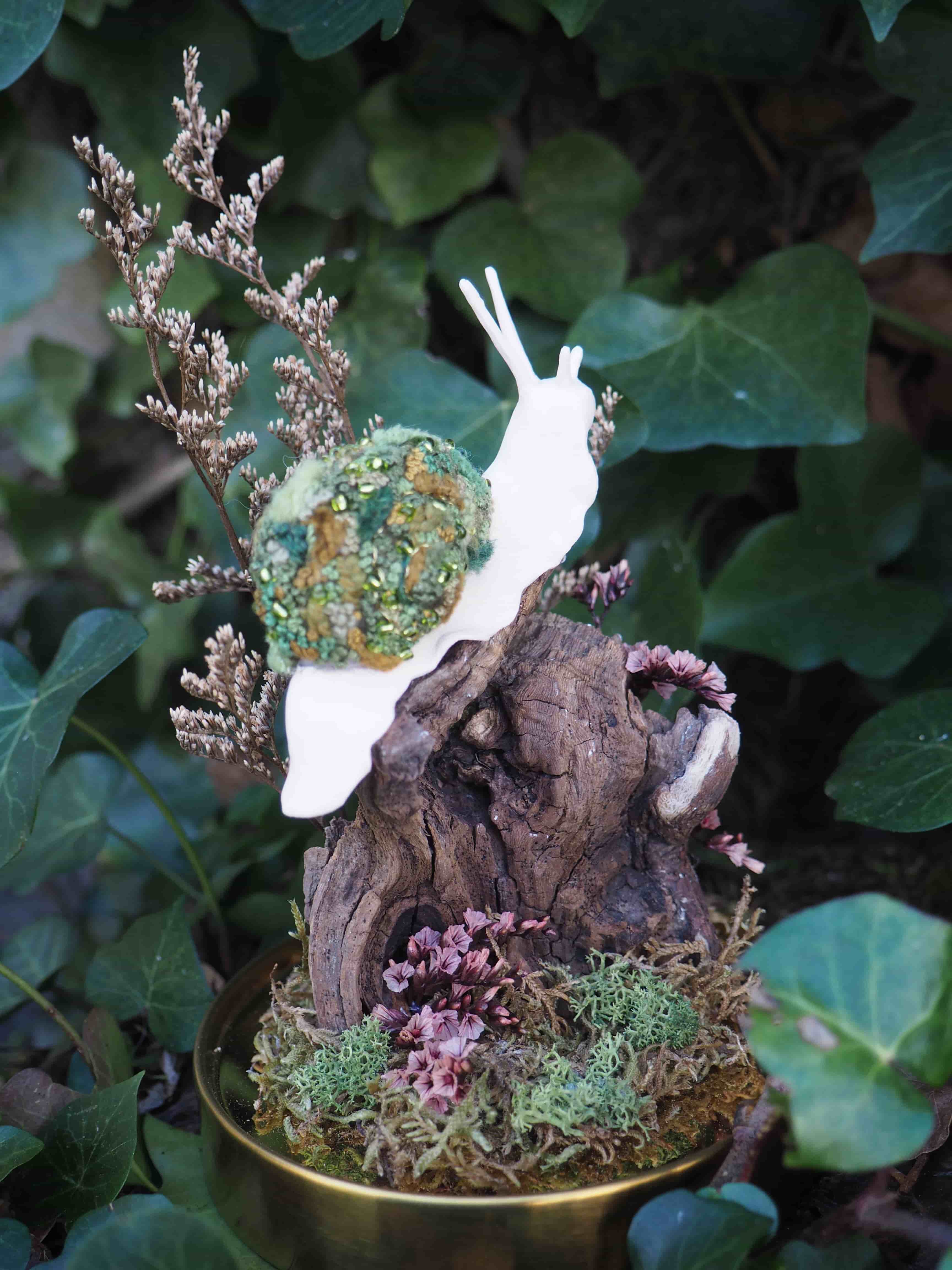 L'image présente une composition artistique et délicate, centrée sur une figurine d'escargot stylisée, posée sur un socle décoratif. Au premier plan, on voit un socle rond et doré, qui contient une petite scène miniature. Sur ce socle, un morceau de bois flotté ou une petite souche d'arbre sert de support principal. Autour de la base du bois, il y a de la mousse verte et de petites fleurs séchées ou des éléments végétaux de couleur rose-violet, créant un petit jardin miniature. L'élément central est une figurine d'escargot. Le corps de l'escargot est d'un blanc pur et lisse, avec des antennes fines et courbées. La coquille de l'escargot est très texturée et colorée, composée de ce qui semble être du feutre ou de la laine cardée dans des tons de vert, de brun et de jaune-doré, créant un motif organique et riche. Derrière la figurine, on aperçoit des branches de fleurs séchées de couleur beige-brun, qui ajoutent de la hauteur et de la texture à la composition. L'arrière-plan est flou, mais on distingue clairement un feuillage vert foncé, probablement du lierre, qui suggère un environnement naturel et extérieur, ou du moins une ambiance de jardin. L'éclairage semble doux et naturel, mettant en valeur les textures et les couleurs de la composition. L'ensemble dégage une atmosphère poétique et artisanale.