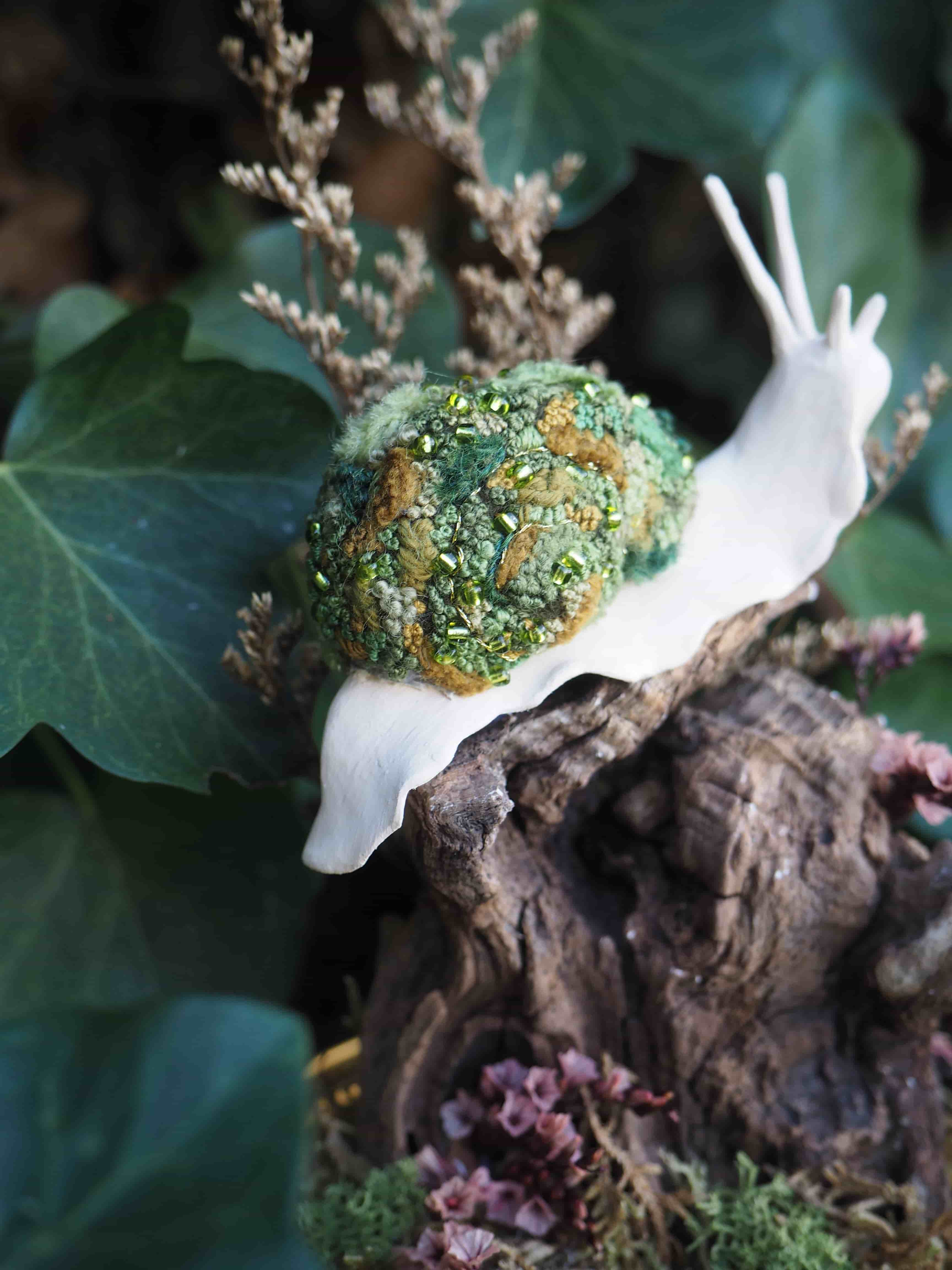 Cette image présente une broche en forme d'escargot, posée sur un morceau de bois flotté ou de souche, dans un environnement qui évoque la nature. L'escargot est l'élément central de l'image. Son corps est d'un blanc immaculé, lisse et mat, contrastant avec sa coquille. La coquille est particulièrement détaillée et texturée, composée de matériaux qui ressemblent à du feutre ou de la laine cardée, dans des nuances de vert et de brun. On y distingue des touches de vert clair, de vert forêt et de brun terreux, avec des petites perles ou des paillettes dorées et vertes incrustées, ajoutant un aspect scintillant et organique, comme de la mousse ou du lichen. Les antennes de l'escargot sont fines et blanches, prolongeant l'élégance de son corps. L'escargot est posé sur un morceau de bois sombre et texturé, qui semble être une souche ou une branche morte, avec des creux et des reliefs. Autour de la base du bois, on aperçoit des éléments végétaux, comme de la mousse verte et des petites fleurs séchées ou des lichens de couleur rosée ou violacée, ajoutant de la profondeur et de la couleur à la scène. En arrière-plan, on distingue un feuillage vert foncé, probablement du lierre, qui crée un fond flou et naturel, mettant en valeur l'escargot. Des éléments végétaux séchés de couleur beige ou marron clair sont également visibles, contribuant à l'ambiance forestière et automnale. La lumière semble douce et naturelle, éclairant l'escargot et ses détails, tout en laissant l'arrière-plan légèrement dans l'ombre, ce qui accentue la mise au point sur l'objet principal. L'ensemble de l'image dégage une atmosphère délicate et artistique, mettant en avant le travail artisanal de la broche.