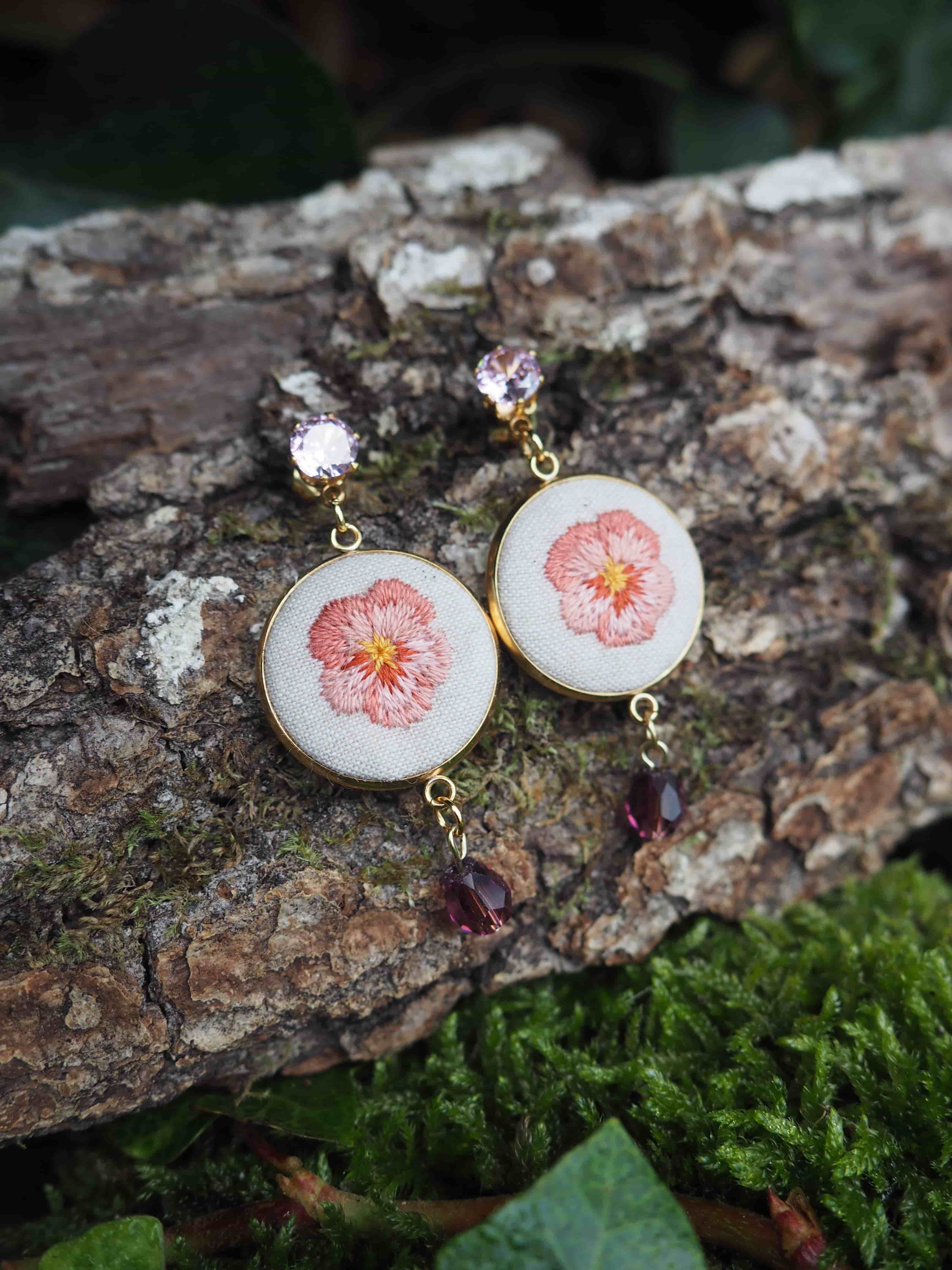 This close-up shot features a pair of elegant dangle earrings, each adorned with a delicate embroidered flower, set against a natural backdrop of tree bark and moss. The earrings are identical in design, each consisting of a circular pendant with a gold-toned frame. Inside each frame, there is a white fabric base on which a small, intricate flower is embroidered. The flower has petals in shades of pink and peach, with a vibrant yellow center, resembling a pansy or viola. Above each circular pendant, a small, faceted, light pink or lavender gemstone is set in a gold-toned claw setting, serving as the stud or top part of the earring. Below each circular pendant, a small, dark red or burgundy teardrop-shaped bead or gemstone dangles, adding a touch of color and movement. The earrings are resting on a piece of rough, textured tree bark, which is dark brown and grey with visible grooves and ridges. To the right and bottom of the bark, there is a patch of bright green moss, providing a soft, natural contrast to the rugged bark. A few dark green leaves are also visible in the foreground and background, further enhancing the organic setting. The lighting is soft and natural, highlighting the details of the embroidery and the sparkle of the gemstones. The focus is sharp on the earrings, with the background slightly blurred to make the jewelry stand out.