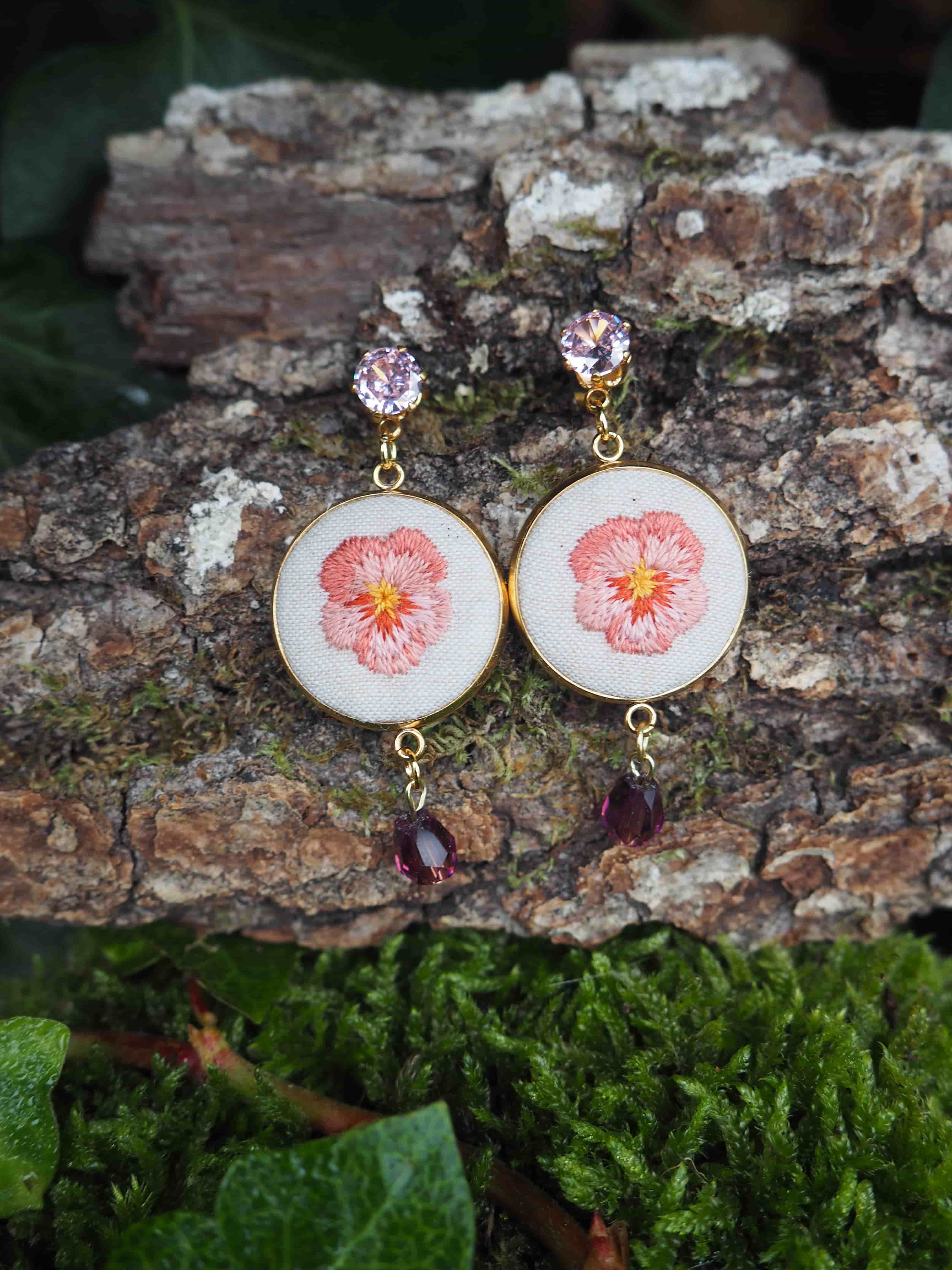 This close-up shot features a pair of handcrafted earrings, each showcasing a delicate embroidered flower, likely a pansy, in shades of pink and orange on a white fabric background. The embroidery is encased in a gold-toned circular frame. Above the embroidered circle, each earring is attached to a small, faceted, light purple or lavender gemstone, which serves as the stud or top part of the earring. Below the embroidered circle, a darker, faceted, teardrop-shaped gemstone, possibly amethyst or a similar purple stone, dangles from a small gold-toned loop. The earrings are displayed against a natural backdrop. They rest on a piece of textured tree bark, which has patches of light green moss growing on it. In the foreground and background, there are hints of dark green foliage, possibly ivy, adding to the organic and rustic aesthetic of the presentation. The lighting appears soft and natural, highlighting the details of the embroidery and the sparkle of the gemstones.