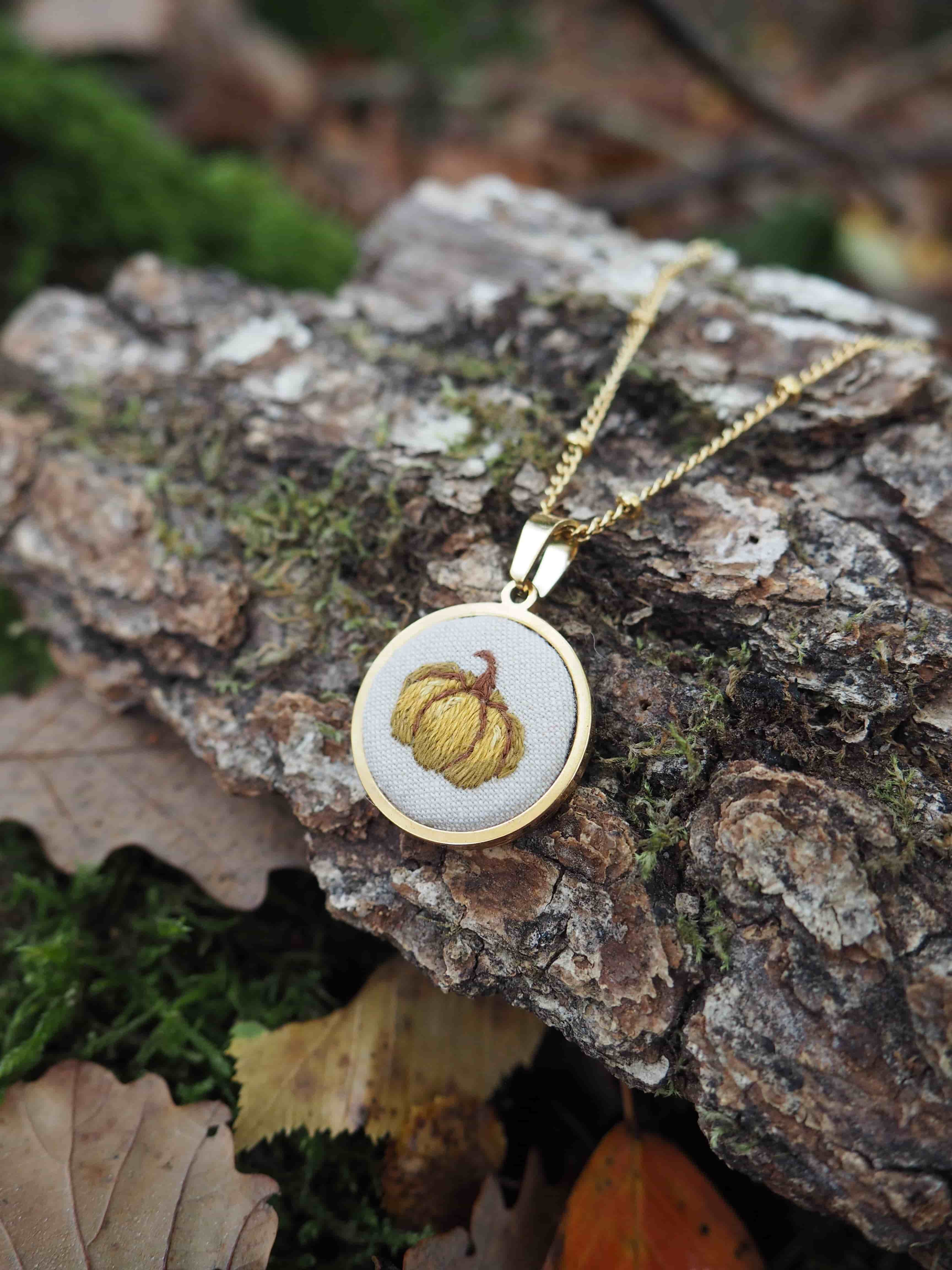 Cette image présente un bijou élégant posé sur un environnement naturel. Il s'agit d'un pendentif circulaire suspendu à une chaîne dorée fine. Le pendentif lui-même arbore un design bicolore avec un cadre doré entourant un centre de couleur claire (probablement blanc ou crème). Au centre du pendentif, on peut observer un motif décoratif qui ressemble à une citrouille ou un potiron stylisé, avec des détails dorés qui évoquent les nervures caractéristiques de ce légume. Le bijou repose sur un morceau d'écorce ou de bois naturel, créant un contraste intéressant entre l'objet manufacturé et l'élément organique. L'arrière-plan révèle un sol forestier avec de la mousse verte, des feuilles mortes et d'autres débris végétaux, suggérant une mise en scène automnale qui s'harmonise parfaitement avec le motif de citrouille du pendentif. L'éclairage naturel met en valeur les détails du bijou tout en créant une atmosphère chaleureuse et saisonnière, évoquant l'automne et ses symboles traditionnels.