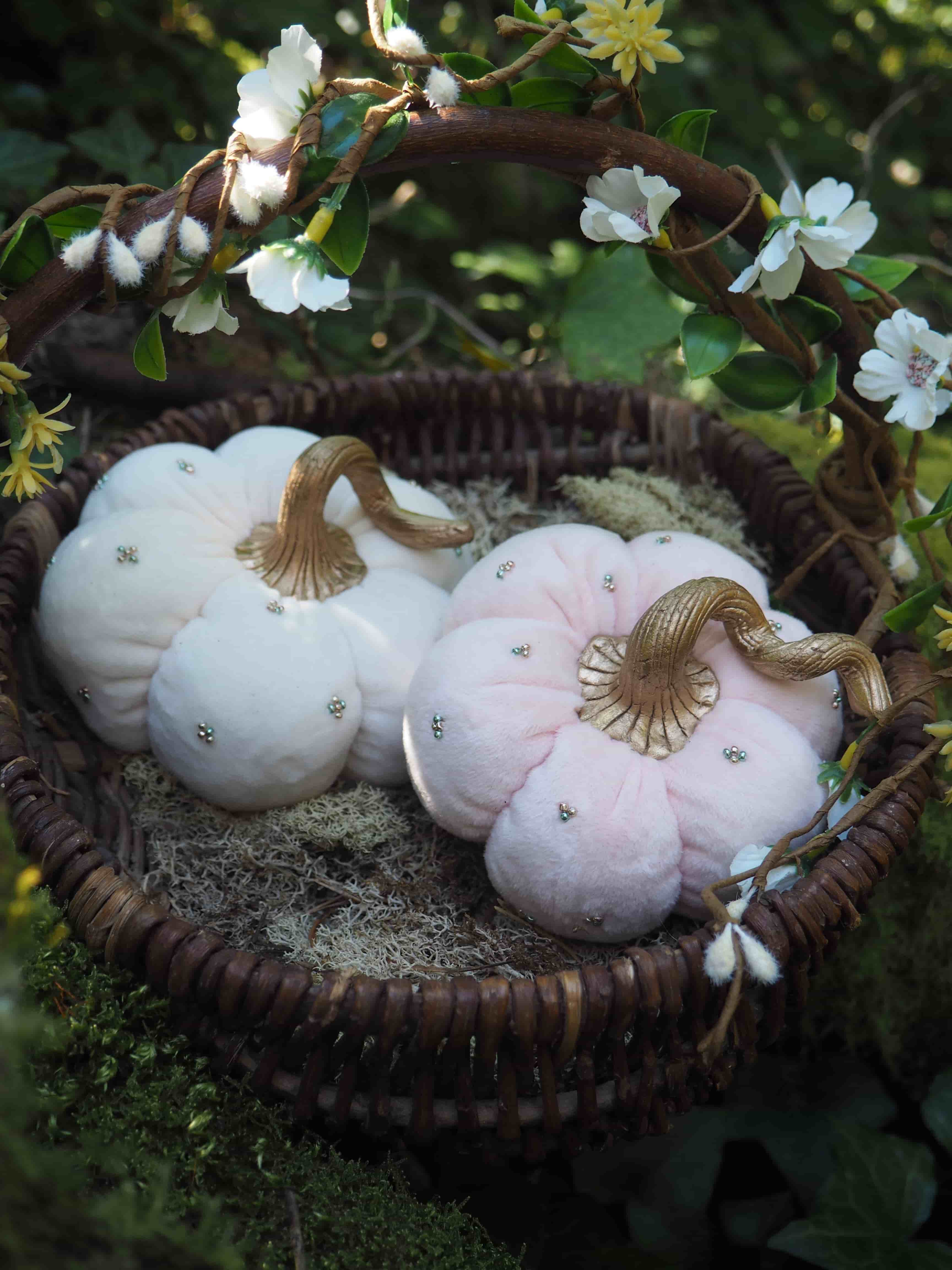 Cette image montre une composition décorative automnale très douce et romantique. Dans un panier en osier foncé sont disposées des citrouilles décoratives en tissu ou en velours, certaines blanches et d'autres dans des tons rose pâle. Ces citrouilles sont ornées de tiges dorées qui leur donnent un aspect élégant. Le panier est placé dans un décor naturel, avec des fleurs blanches délicates (qui semblent être des fleurs de pommier ou de cerisier) encadrant la scène. L'ensemble crée une ambiance très poétique et féerique, s'éloignant du style traditionnel orange et noir d'Halloween pour offrir une décoration plus douce et raffinée.