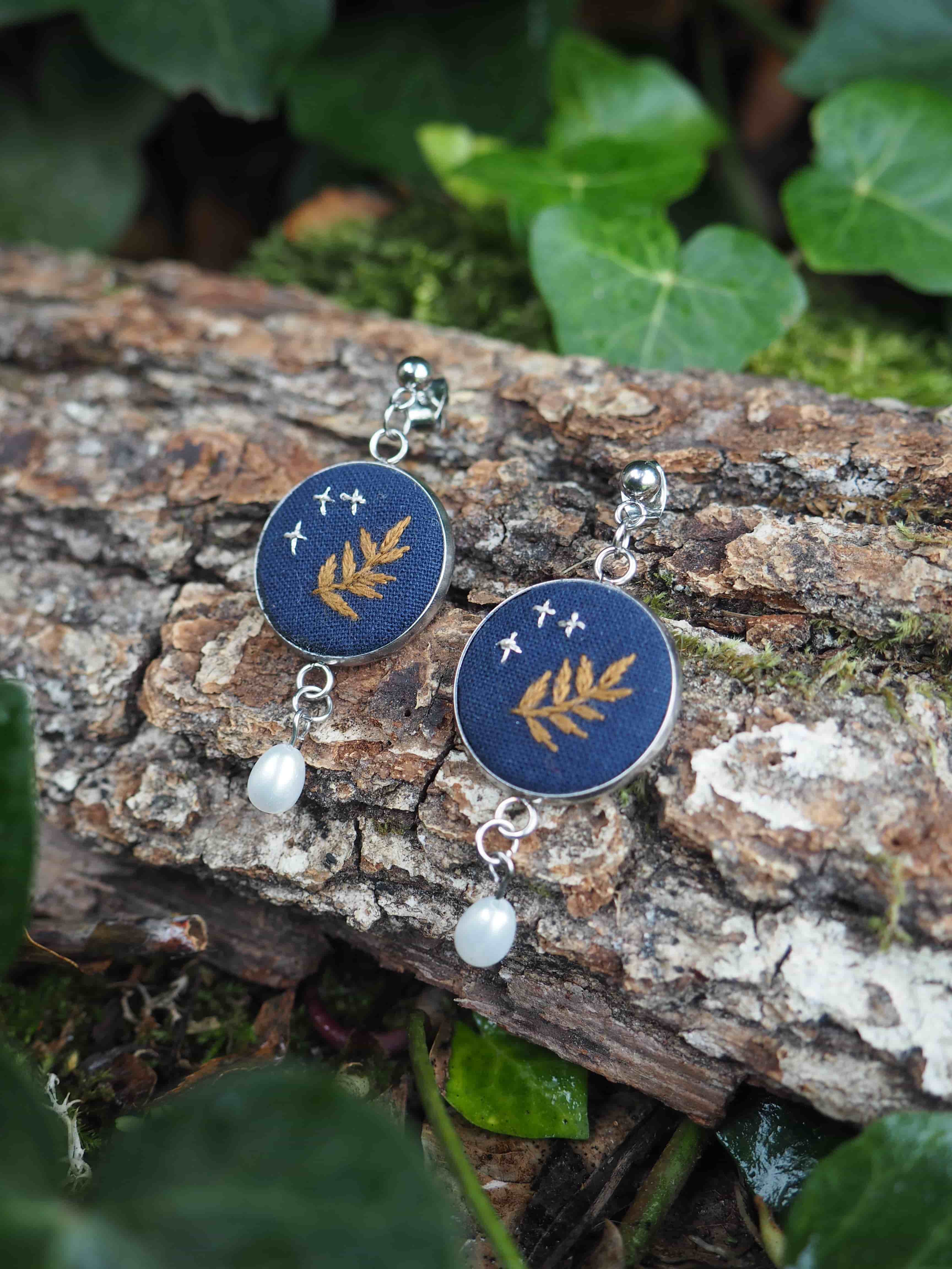 Cette image présente une paire de boucles d'oreilles artisanales disposées sur un morceau de bois naturel dans un environnement végétal. **Description des bijoux :**
- Boucles d'oreilles avec des pendentifs circulaires de couleur bleu foncé/marine
- Chaque pendentif est orné d'un motif doré représentant une feuille ou une branche délicate
- De petites étoiles dorées parsèment le fond bleu, créant un effet céleste
- Une perle blanche nacrée pend élégamment sous chaque pendentif principal
- Les attaches et chaînettes sont en métal argenté **Mise en scène :**
- Les bijoux reposent sur une branche ou un tronc d'arbre à l'écorce rugueuse et naturelle
- L'arrière-plan est composé de feuillage vert luxuriant, légèrement flou
- L'éclairage naturel met en valeur les détails dorés et la brillance des perles
- Cette présentation évoque un style bohème et naturel, soulignant le caractère artisanal des pièces L'ensemble dégage une esthétique poétique mêlant thèmes botaniques et cosmiques, parfaite pour des bijoux de créateur inspirés de la nature.