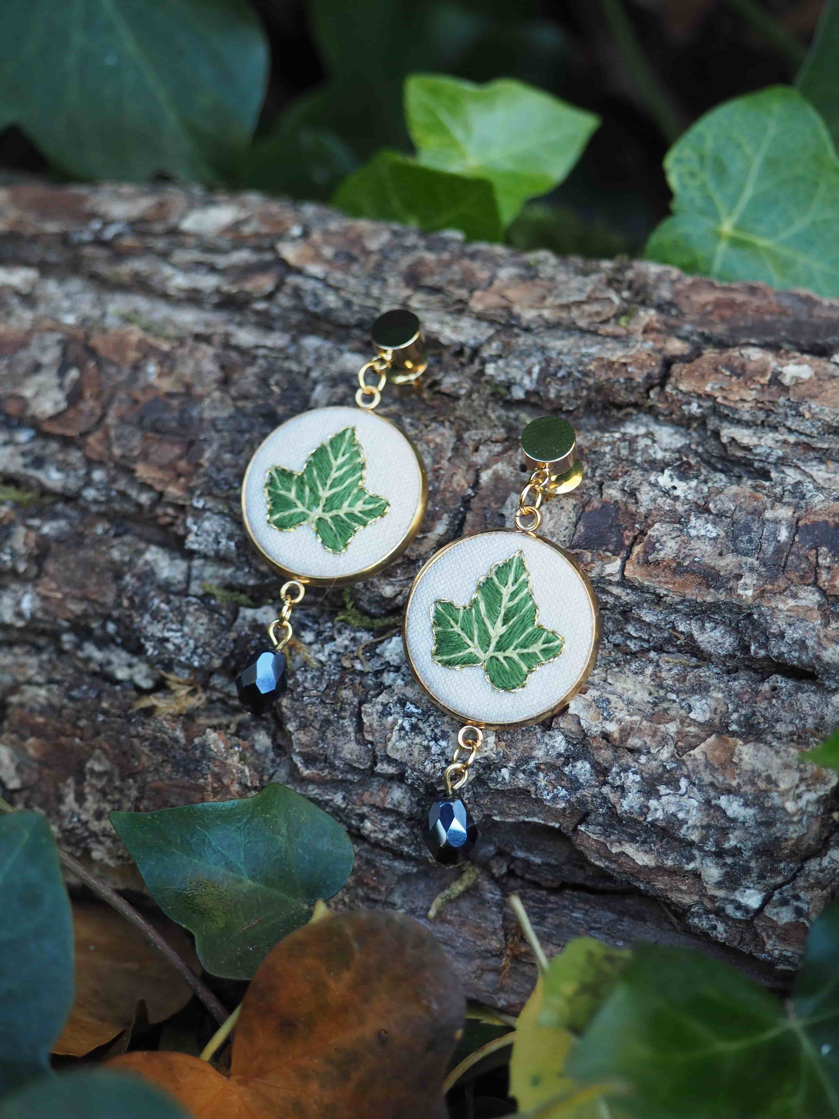 Cette image présente une paire de boucles d'oreilles artisanales disposées sur un tronc d'arbre dans un environnement naturel.  **Caractéristiques des bijoux :**
- Pendants circulaires en céramique ou porcelaine blanche
- Motifs de feuilles vertes peints à la main avec des détails délicats
- Montures dorées élégantes
- Petites perles bleues suspendues en bas de chaque boucle d'oreille
- Style bohème et naturel **Mise en scène :**
- Les bijoux reposent sur l'écorce rugueuse et texturée d'une branche
- Environnement forestier avec des feuilles vertes floues en arrière-plan
- Éclairage naturel doux qui met en valeur les couleurs
- Composition artistique qui crée une harmonie entre les bijoux et leur environnement naturel L'ensemble évoque un style artisanal raffiné, parfait pour quelqu'un qui apprécie les accessoires inspirés de la nature avec une touche d'élégance.
