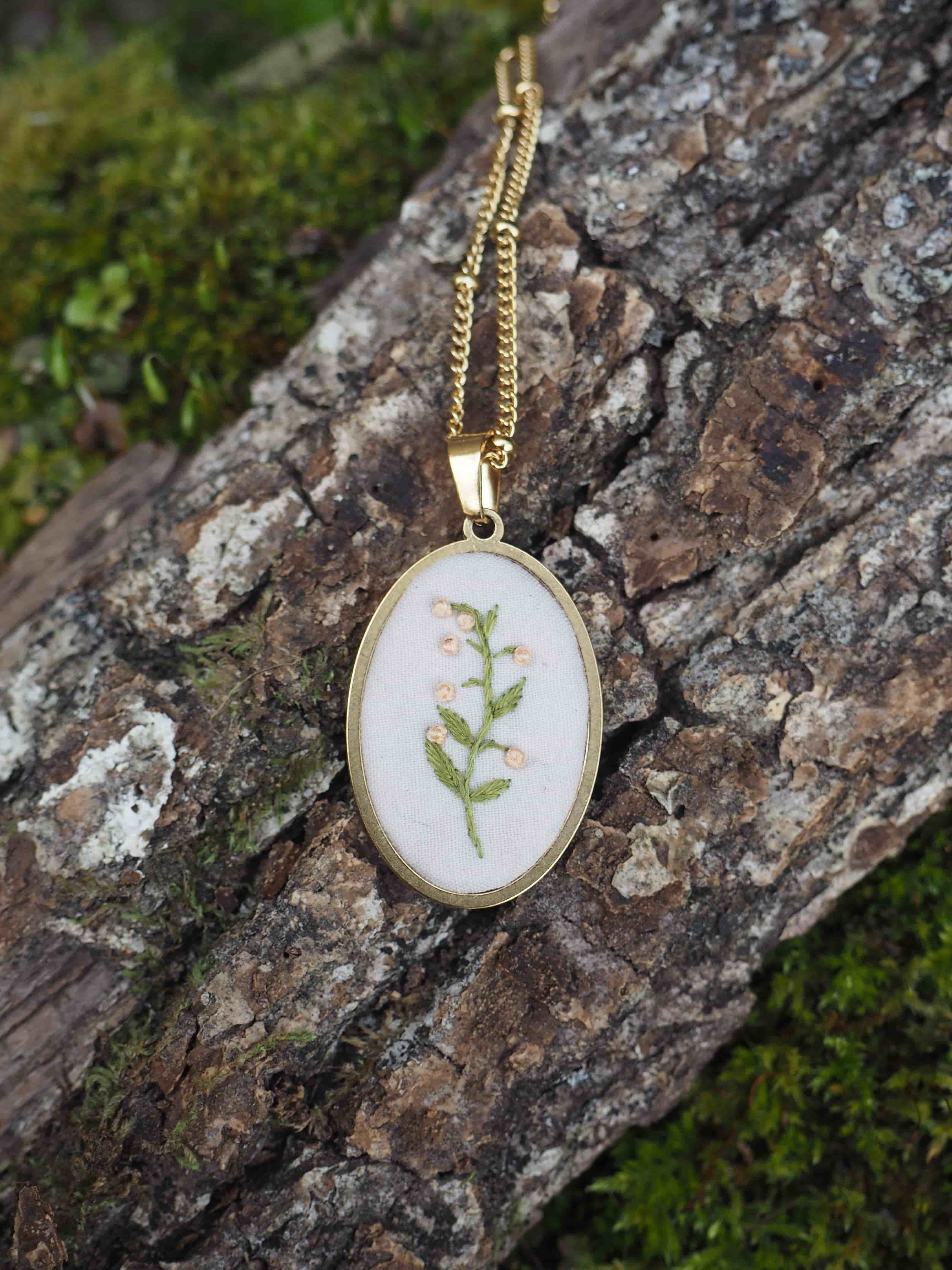 Cette image montre un délicat pendentif artisanal sur une chaîne dorée. Le pendentif est composé d'un cercle en métal doré qui encadre une broderie ou une impression botanique sur fond blanc. Le motif représente une petite fougère ou une brindille végétale verte, créant un effet naturel et délicat. Le bijou est photographié sur ce qui semble être un morceau de bois ou d'écorce recouvert de mousse, ce qui crée une mise en scène naturelle qui complimente bien le thème botanique du pendentif. L'ensemble dégage une atmosphère naturelle et poétique.