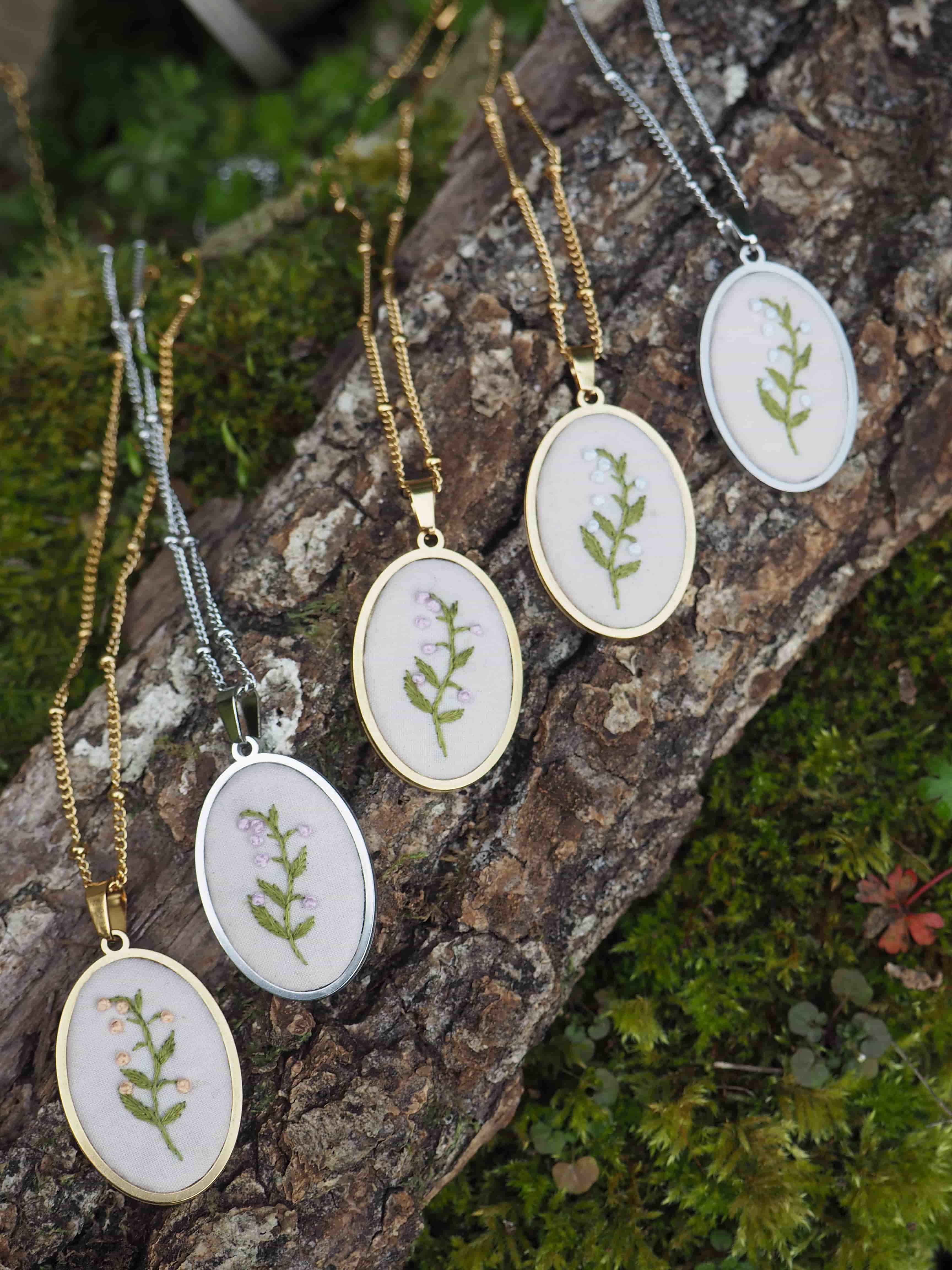 Cette image montre une collection de délicats pendentifs brodés. Chaque pendentif est composé d'un petit cercle à broderie contenant un motif végétal simple - une petite branche verte avec des feuilles - encadré dans un support métallique rond. Les pendentifs sont présentés sur ce qui semble être un morceau d'écorce d'arbre ou de bois recouvert de mousse, créant un cadre naturel et organique. Les chaînes sont disponibles en deux finitions : dorée et argentée. L'ensemble dégage une atmosphère naturelle et botanique, parfaite pour les amateurs de bijoux inspirés par la nature.