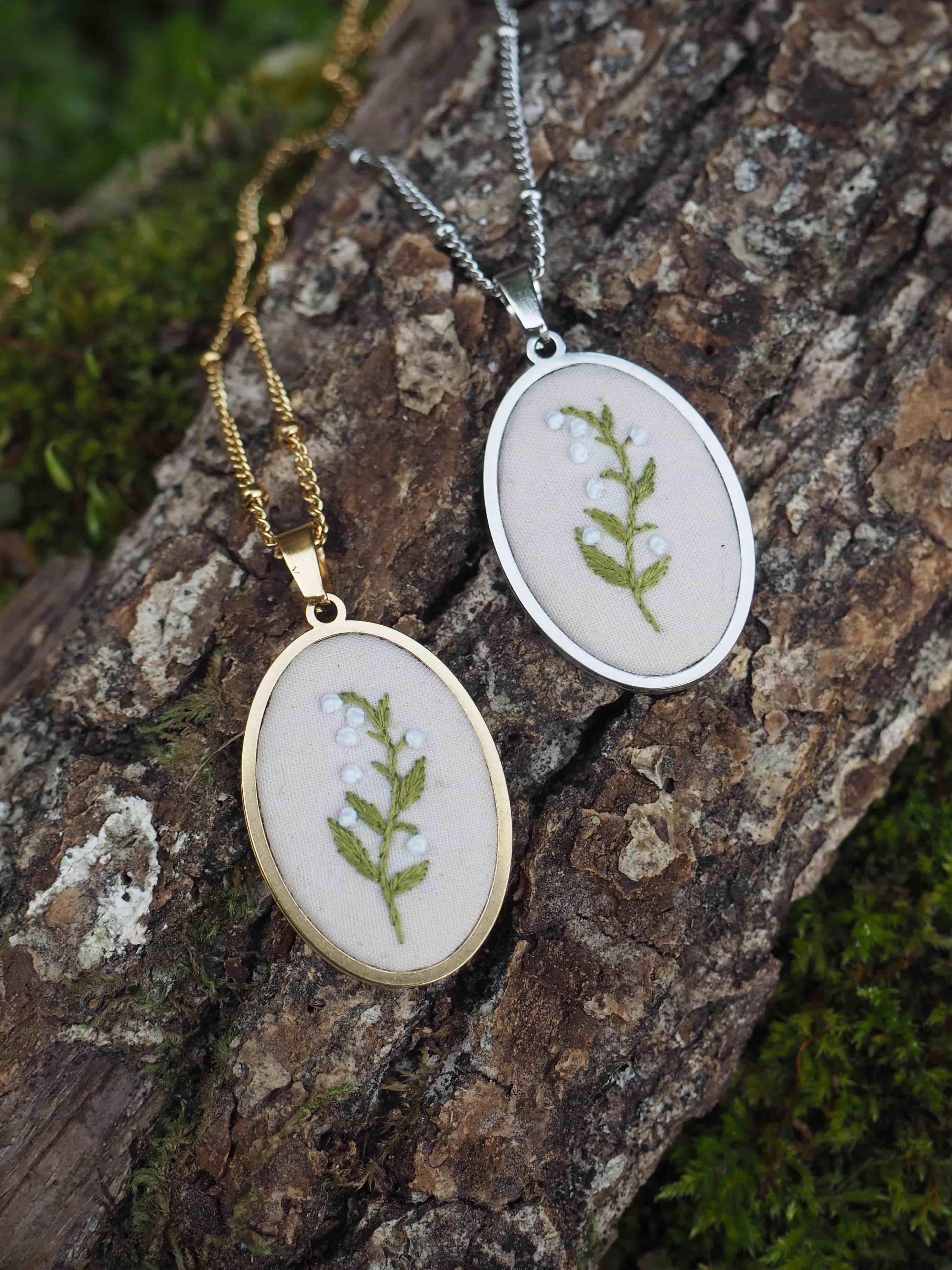 Cette image montre deux pendentifs délicats posés sur un morceau d'écorce d'arbre moussue. Les pendentifs sont de forme ovale, l'un en finition dorée et l'autre en finition argentée. Chacun contient une petite fougère ou une feuille verte pressée et préservée sous ce qui semble être du verre ou une résine transparente. Les pendentifs sont suspendus à des chaînes fines assorties à leur couleur respective (dorée et argentée). C'est un bel exemple de bijoux naturels qui incorporent des éléments botaniques, créant une pièce élégante et organique. La texture rugueuse de l'écorce et la mousse verte en arrière-plan complètent parfaitement ces bijoux nature.
