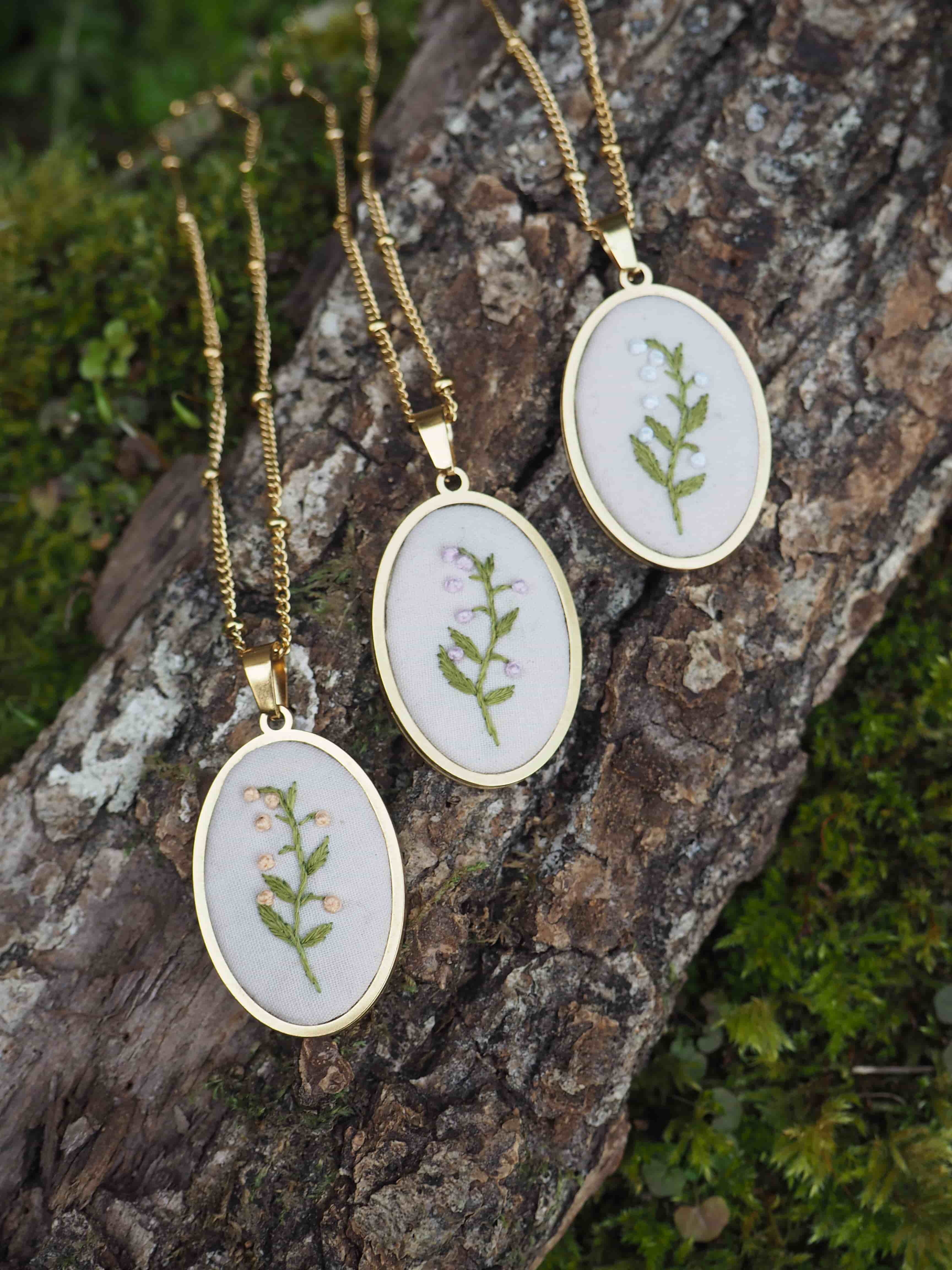 Cette image montre trois délicats pendentifs identiques disposés sur un tronc d'arbre couvert de mousse. Chaque pendentif est composé d'un cercle blanc brodé avec un motif de feuille ou de brindille verte, encadré dans un support doré. Les pendentifs sont attachés à des chaînes dorées fines. Le contraste entre les pendentifs clairs et la texture sombre et rugueuse de l'écorce d'arbre crée une belle composition naturelle. Ces bijoux semblent être des pièces artisanales qui incorporent des éléments botaniques dans leur design, ce qui est très tendance dans la bijouterie contemporaine.