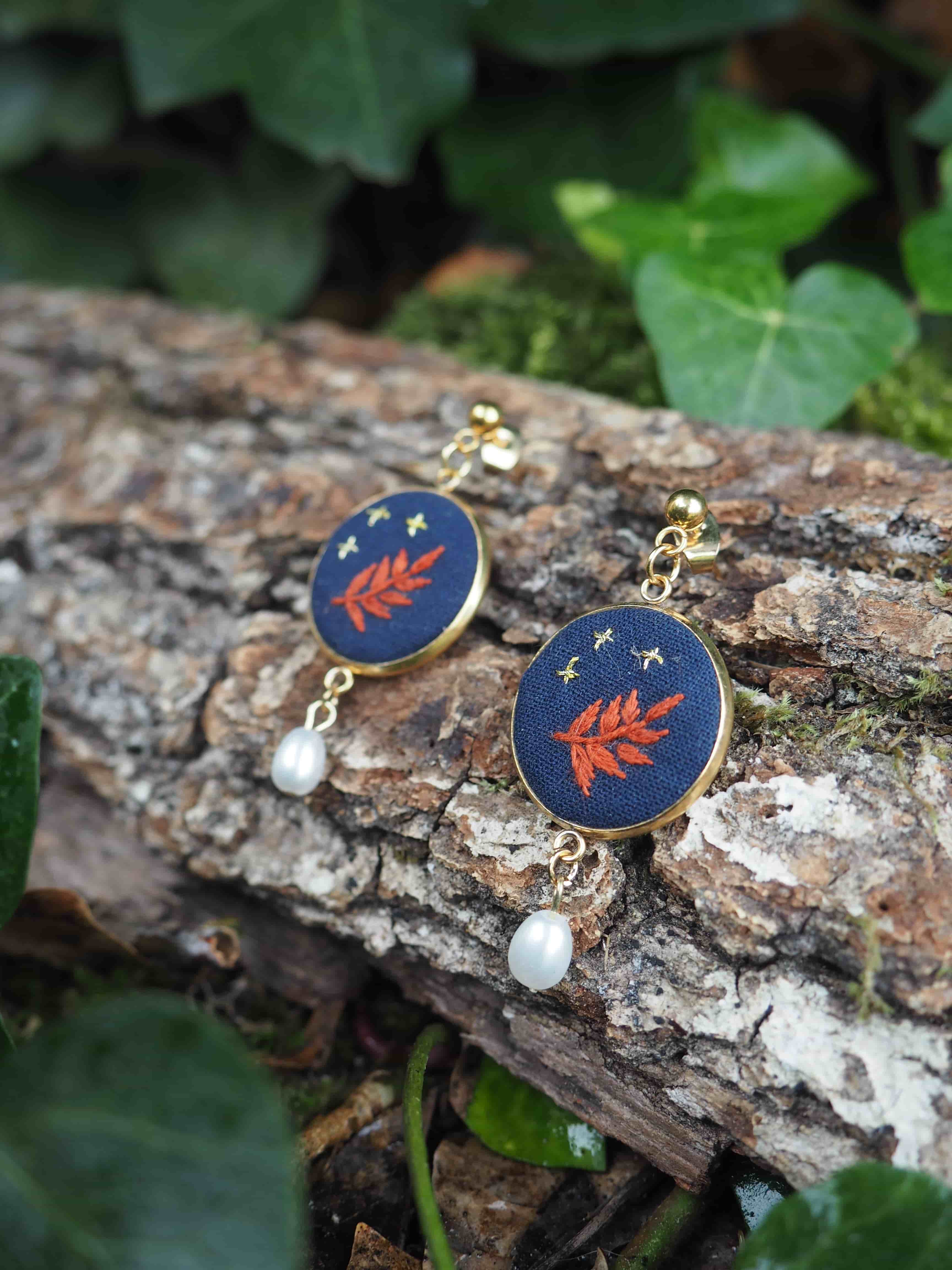 Cette image présente une paire de boucles d'oreilles élégantes posées sur un morceau de bois naturel dans un environnement verdoyant. **Description des bijoux :**
- Boucles d'oreilles composées de médaillons circulaires à fond bleu marine
- Motif décoratif : une feuille ou branche stylisée de couleur rouge-orangé avec de petites étoiles dorées dispersées
- Monture dorée qui encadre chaque médaillon
- Petites perles blanches nacrées suspendues en pendentif sous chaque médaillon
- Style raffiné mêlant couleurs vives et éléments naturels **Mise en scène :**
- Les bijoux reposent sur une surface de bois brut, avec une écorce texturée et patinée
- Arrière-plan composé de feuillage vert luxuriant, légèrement flou
- Éclairage naturel qui met en valeur les couleurs et les détails des boucles d'oreilles
- Composition harmonieuse entre les éléments naturels et les bijoux artisanaux L'ensemble crée une atmosphère organique et artisanale, suggérant des bijoux de créateur inspirés par la nature.