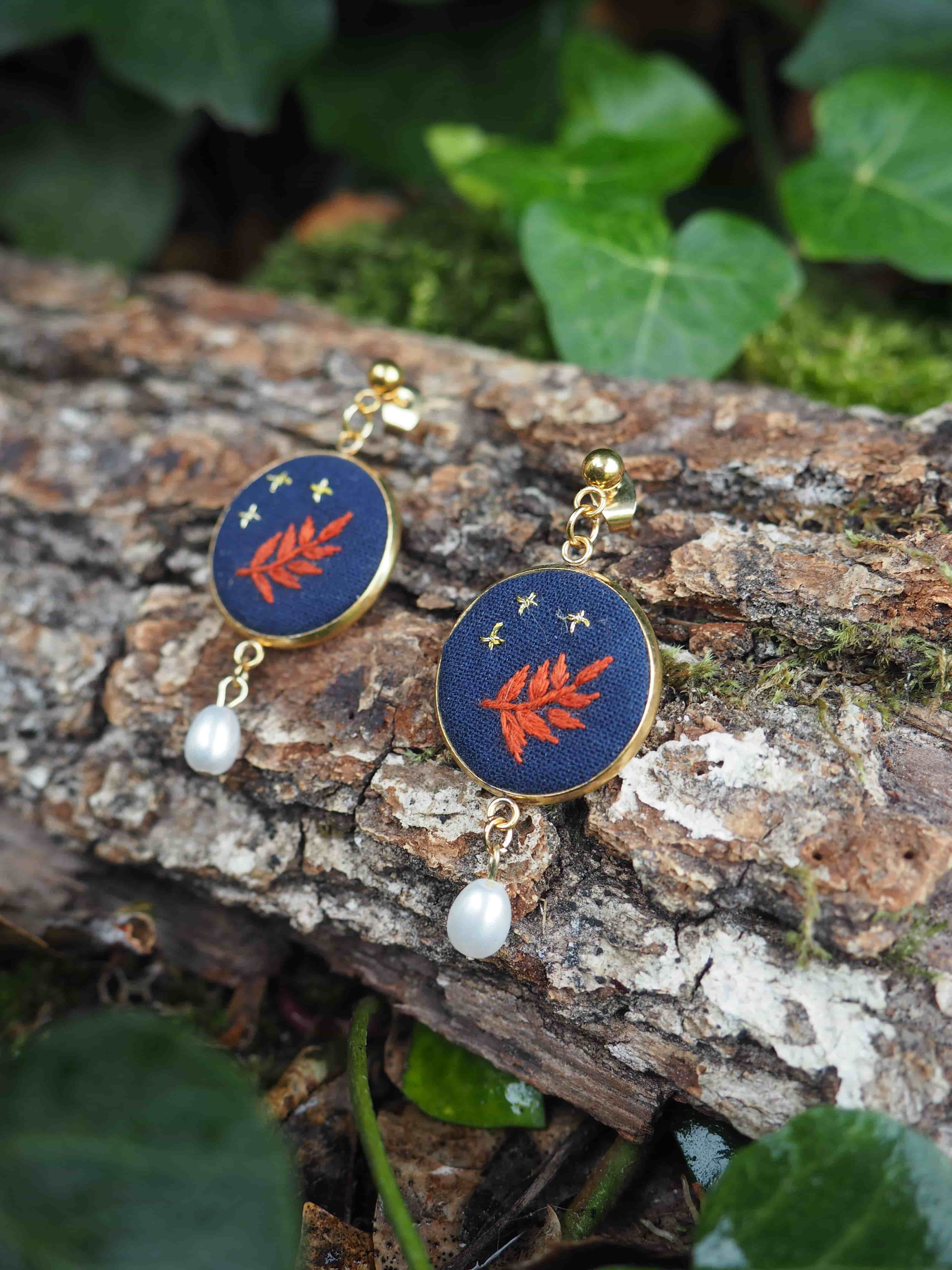 Cette image présente une paire de boucles d'oreilles artisanales disposées sur un tronc d'arbre dans un environnement naturel.  **Caractéristiques des bijoux :**
- Forme circulaire avec un fond bleu marine/navy
- Motif central représentant une feuille ou branche stylisée en couleur orange/corail
- Petites étoiles dorées dispersées sur le fond bleu
- Perle blanche nacrée suspendue à chaque boucle
- Crochets dorés pour l'attache **Environnement :**
- Les boucles d'oreilles reposent sur l'écorce rugueuse d'un tronc d'arbre
- Arrière-plan naturel avec un feuillage vert luxuriant légèrement flou
- Éclairage naturel qui met en valeur les couleurs et les détails
- Quelques traces de lichen ou mousse visible sur l'écorce Le style de ces bijoux évoque un design bohème ou artisanal, avec une esthétique qui rappelle un ciel étoilé nocturne orné d'éléments botaniques. La mise en scène naturelle renforce le caractère organique et authentique de ces accessoires.