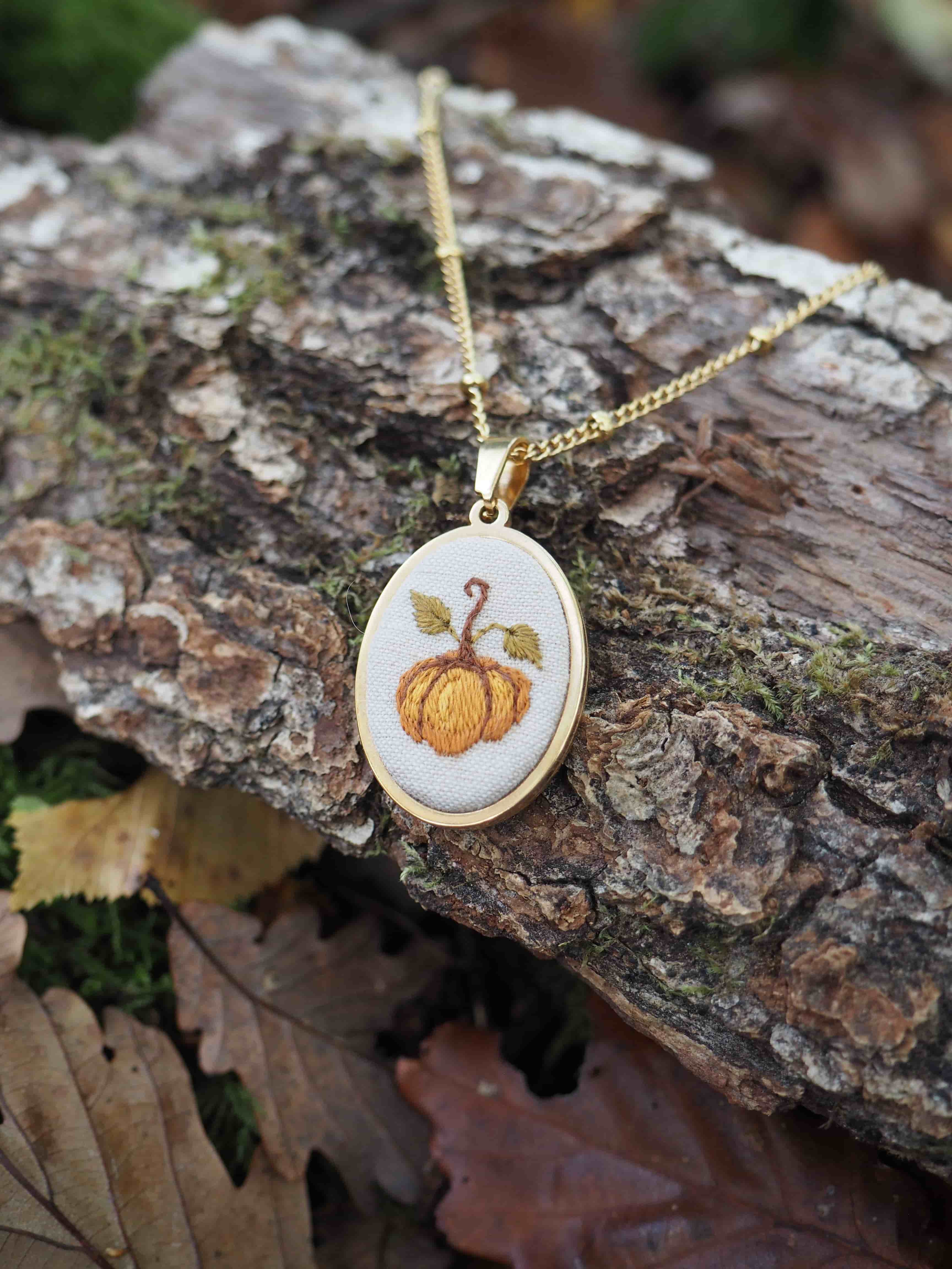 Cette image présente un joli collier artisanal posé sur un morceau d'écorce d'arbre dans un environnement naturel. Le pendentif est constitué d'un petit cerceau en bois clair (probablement du bouleau ou un bois similaire) dans lequel est brodé un motif de citrouille orange avec une tige verte et des feuilles. Cette broderie évoque l'esprit automnal et la saison d'Halloween. Le collier est suspendu à une chaîne dorée délicate. L'ensemble repose sur un morceau d'écorce rugueuse et texturée, entouré de feuilles mortes aux tons bruns et dorés, ce qui renforce l'atmosphère automnale de la composition. L'éclairage naturel met en valeur les détails de la broderie et crée une ambiance chaleureuse. Cette pièce de bijouterie artisanale semble être un accessoire parfait pour célébrer la saison d'automne, alliant savoir-faire traditionnel de la broderie et esthétique naturelle.