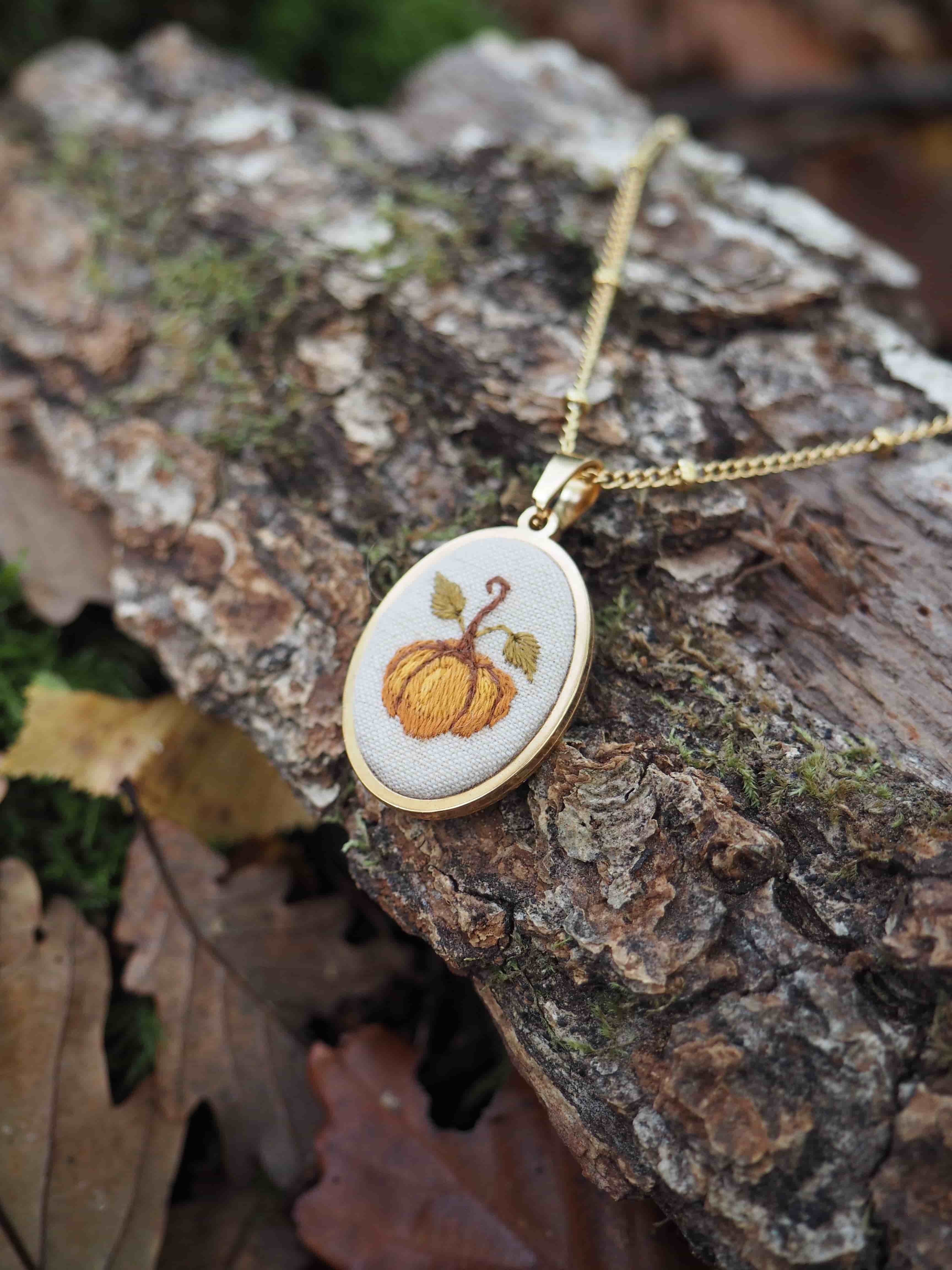 Cette image présente un délicat pendentif artisanal posé sur un morceau d'écorce d'arbre dans un environnement naturel. Le bijou se compose d'un petit cadre ovale en bois clair, probablement du bouleau ou un bois similaire, suspendu à une fine chaîne dorée. À l'intérieur du pendentif, on peut admirer une broderie minutieuse représentant une citrouille orange avec sa tige verte, réalisée sur un tissu de fond blanc ou crème. Les détails de la broderie sont particulièrement soignés, avec des nuances d'orange qui donnent du relief et de la texture au motif automnal. Le pendentif repose sur une surface d'écorce rugueuse et texturée, parsemée de mousse verdâtre, ce qui crée un contraste intéressant entre l'objet délicat et l'environnement naturel brut. En arrière-plan, on distingue des feuilles mortes aux tons bruns et cuivrés, renforçant l'atmosphère automnale de la composition. L'ensemble évoque un style artisanal fait main, probablement une pièce unique alliant broderie traditionnelle et joaillerie contemporaine, parfaite pour célébrer la saison d'automne.