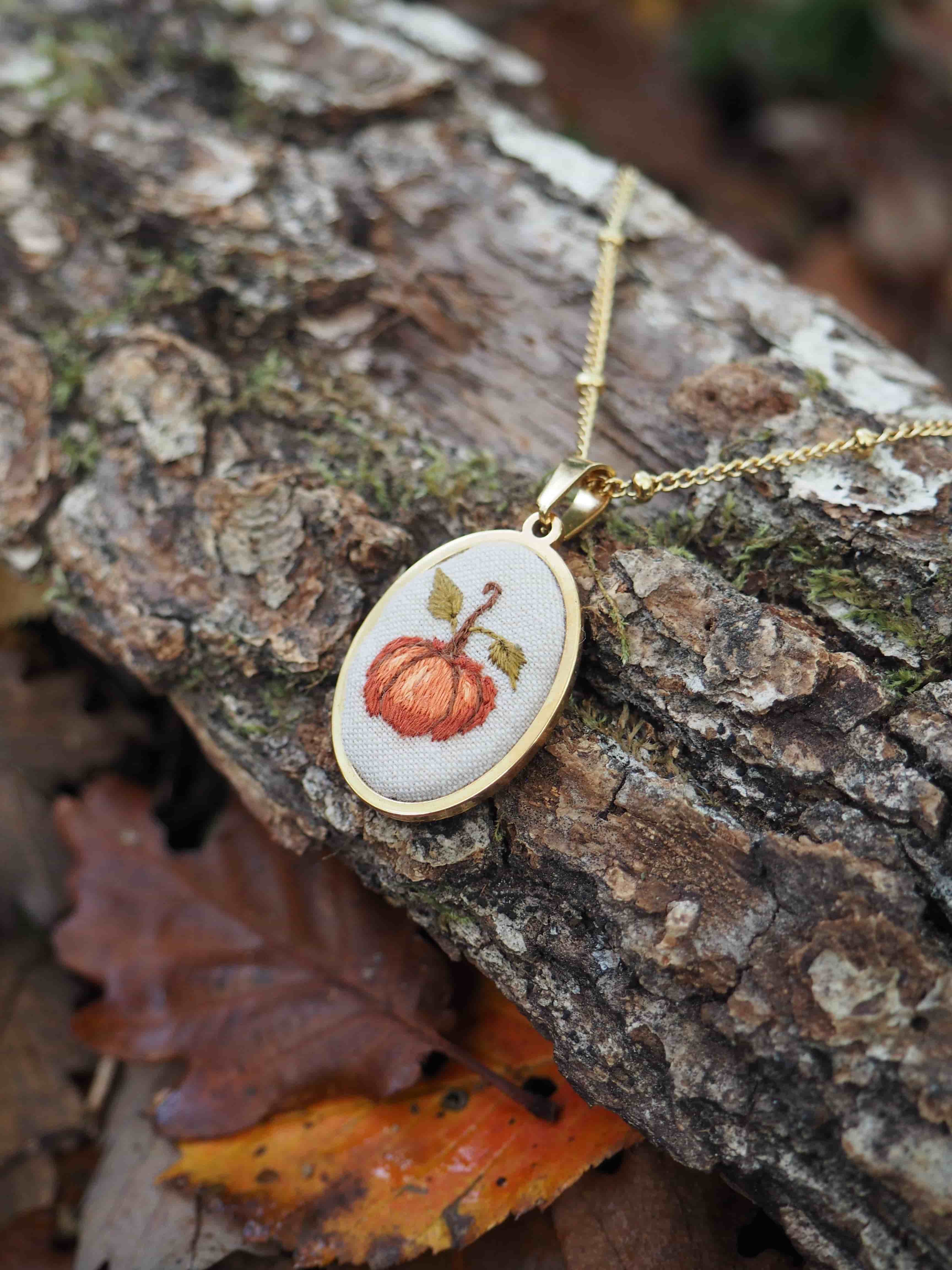 Cette image présente un délicat pendentif artisanal posé sur un morceau d'écorce d'arbre dans un environnement naturel automnal. Le bijou se compose d'un petit cadre ovale doré contenant une broderie minutieuse représentant une citrouille orange avec sa tige verte. Le travail de broderie est remarquablement détaillé, avec des points soignés qui donnent du relief et de la texture au motif. Le pendentif est suspendu à une fine chaîne dorée et repose sur l'écorce rugueuse et texturée d'un tronc d'arbre, dont on peut voir les nuances de brun et les traces de mousse. En arrière-plan, on aperçoit des feuilles mortes aux teintes automnales - orange, brun et jaune - qui jonchent le sol, créant une atmosphère saisonnière parfaitement en harmonie avec le motif de citrouille du bijou. L'ensemble évoque l'artisanat fait main, la saison d'automne et la beauté des petits détails décoratifs inspirés de la nature. La composition photographique met en valeur le contraste entre la délicatesse du travail de broderie et la rusticité de l'environnement naturel.