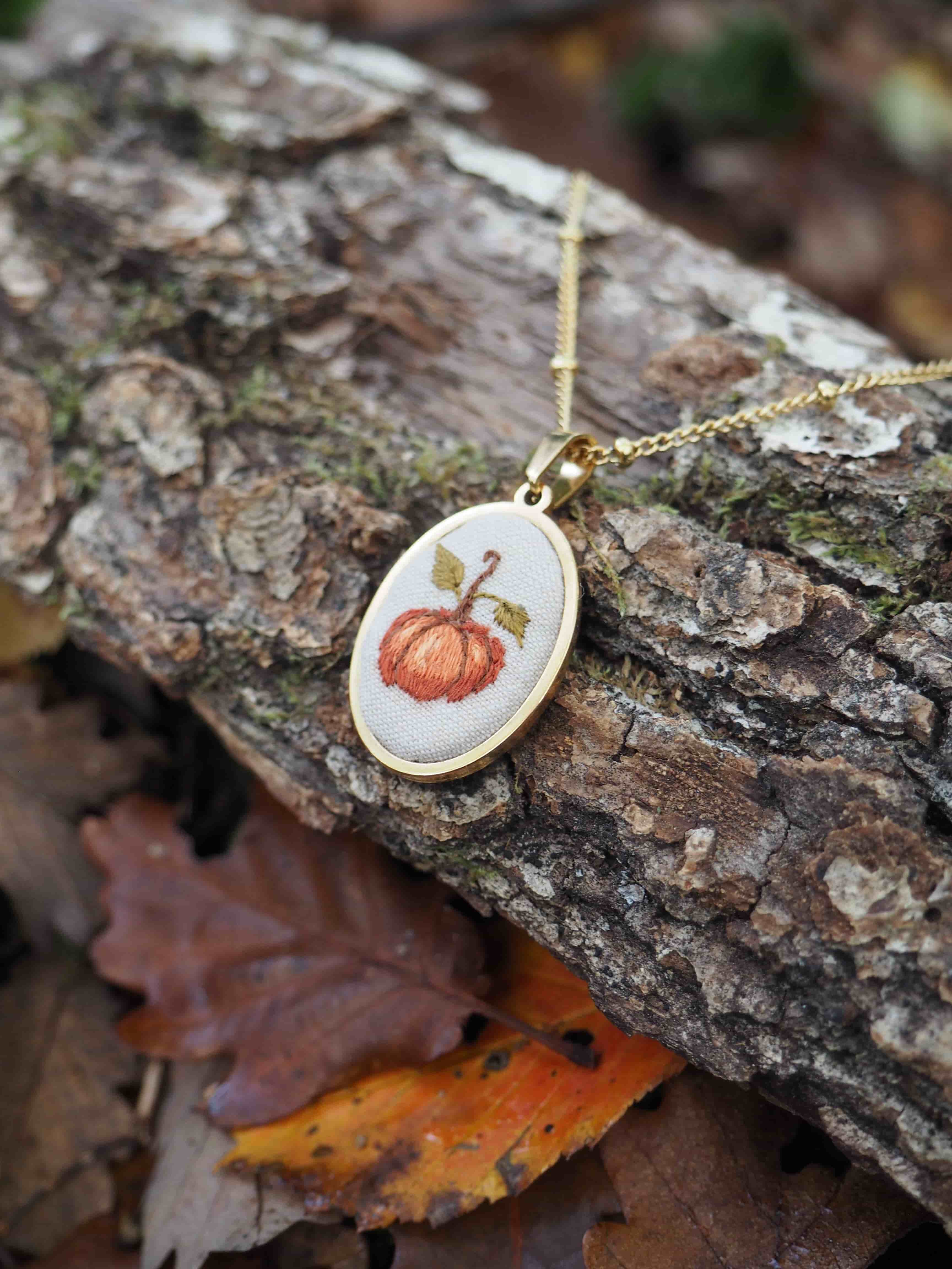 Cette image présente un délicat pendentif artisanal posé sur un morceau d'écorce d'arbre dans un environnement naturel automnal. Le bijou se compose d'un médaillon ovale à bordure dorée, suspendu à une fine chaîne également dorée.  Au centre du pendentif, on découvre une broderie minutieuse représentant une citrouille orange avec sa tige verte, réalisée sur un fond de tissu blanc ou crème. Cette petite œuvre d'art textile évoque parfaitement l'esprit de l'automne et d'Halloween. L'écorce sur laquelle repose le bijou présente une texture rugueuse et naturelle, typique d'un tronc d'arbre, avec ses crevasses et ses nuances de brun. Autour, on aperçoit quelques feuilles mortes aux teintes automnales - orange et brun - qui complètent harmonieusement la composition et renforcent l'atmosphère saisonnière de la photographie. L'ensemble crée une mise en scène poétique qui met en valeur ce bijou fait main aux accents champêtres et festifs.