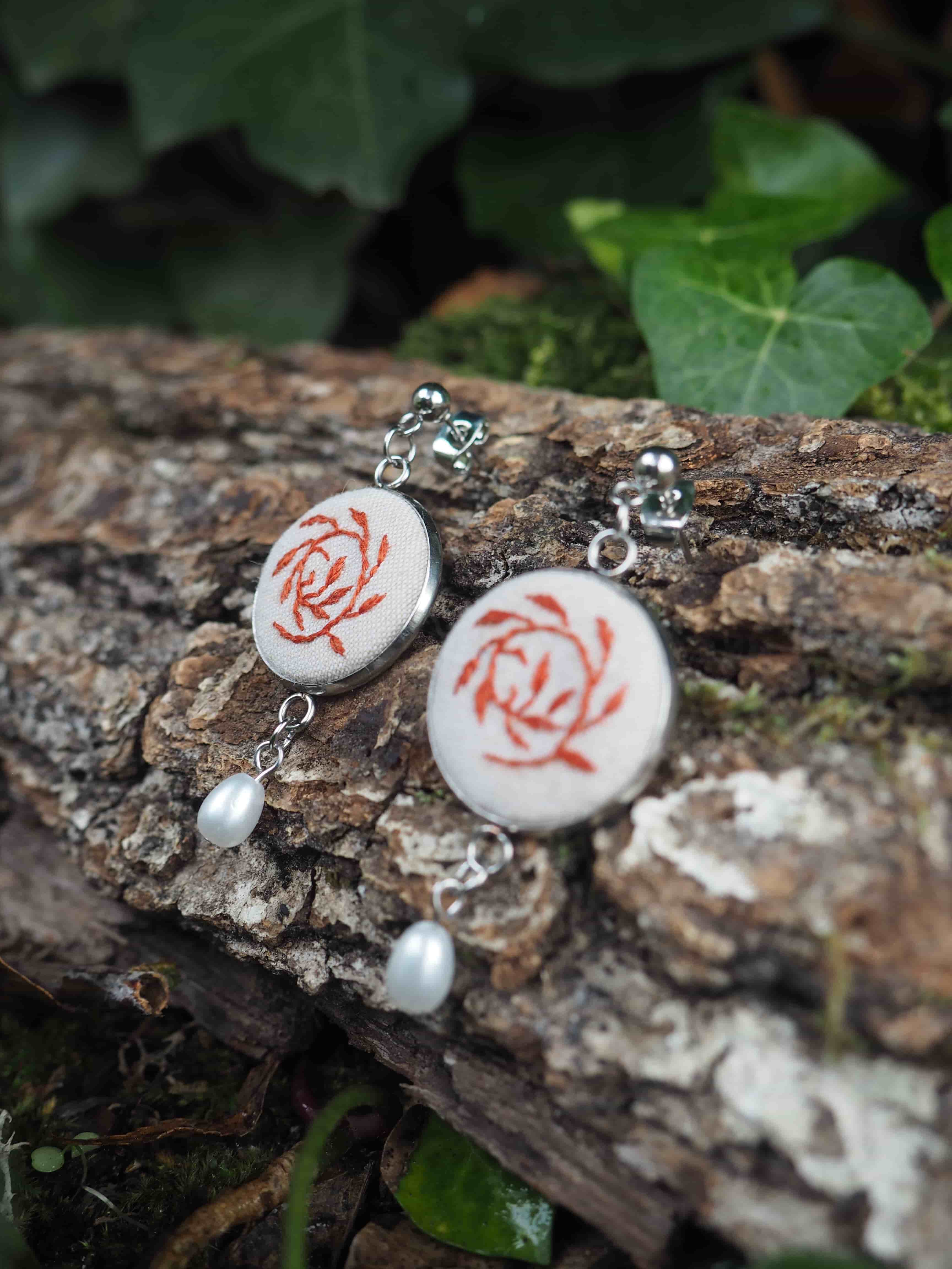 Cette image présente une paire de boucles d'oreilles artisanales disposées sur un tronc d'arbre dans un environnement naturel.  **Caractéristiques des bijoux :**
- Pendants circulaires en céramique ou porcelaine blanche
- Motif floral rouge/rose représentant des roses stylisées
- Montures en métal argenté avec des crochets d'oreilles
- Petites perles blanches nacrées suspendues en pendentif
- Style romantique et délicat **Mise en scène :**
- Posés sur l'écorce rugueuse et texturée d'un tronc d'arbre
- Arrière-plan naturel avec un feuillage vert flou
- Éclairage naturel doux qui met en valeur les détails
- Composition photographique soignée créant un contraste entre la délicatesse des bijoux et la rusticité du support en bois L'ensemble évoque un style artisanal fait main, avec une esthétique à la fois naturelle et raffinée, parfait pour un look bohème ou romantique.